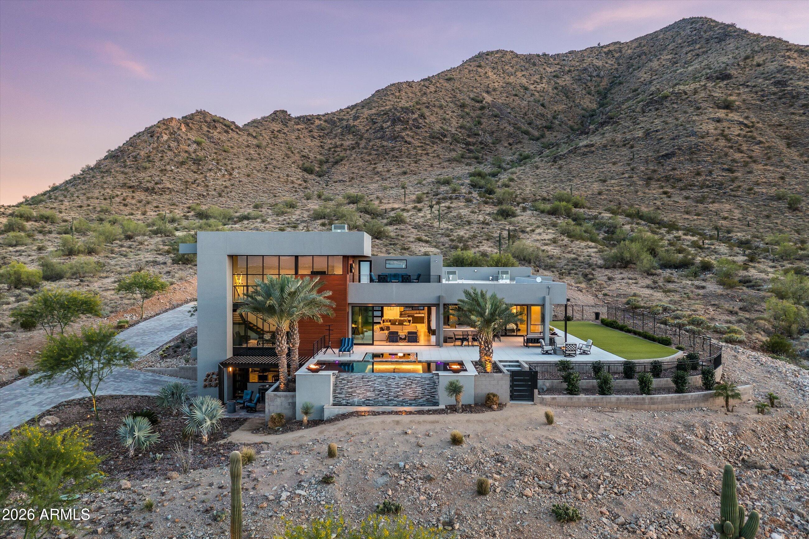 Welcome to The Lions Gate, a striking desert estate set on 3.87 acres of pristine Sonoran landscape. Designed to fully engage its surroundings, the home is positioned to capture sweeping views, expansive skies, and a level of privacy that feels both rare and intentional.
From the moment of arrival, bold architectural lines and expansive walls of glass establish a dialogue between structure and setting. The desert is not simply outside the home, it becomes part of the experience within. Light moves throughout the interiors, shifting with the time of day and softening each space with a quiet, natural rhythm. The result is a residence that feels grounded, open, and deeply connected to place.
At the center of the home, the kitchen unfolds as a natural gathering space, seamlessly connected to to the main living areas. Designed with both function and atmosphere in mind, it supports intimate daily living as effortlessly as it does larger moments of entertaining. Custom finishes and thoughtful detailing create a sense of cohesion, where each element contributes to the overall composition of the home.
The primary suite exists as a private retreat, carefully positioned to take in the surrounding landscape. Views extend outward in every direction, creating a sense of stillness and separation from the rest of the home. Additional spaces offer flexibility for guests, a home office, or wellness, adapting easily to the needs of daily life.
Outdoors, the property unfolds into a series of expansive living environments shaped by the desert itself.  Patios extend into the landscape, offering multiple vantage points to experience the changing light and atmosphere throughout the day. In the evening, the setting transforms as the sky deepens into color, revealing layered sunsets over the mountains and a soft emergence of city lights in the distance. Mountain vistas frame the horizon, while the surrounding acreage preserves a sense of openness and quiet that is increasingly difficult to find.
Here, outdoor living is not an extension of the home, it is an essential part of it. Each space is positioned to invite pause, to capture view, and to create a connection to the natural surroundings.
The Lions Gate is more than a residence. It is a composition of land, light, and perspective, brought together to create a living experience defined by scale, privacy, and presence.