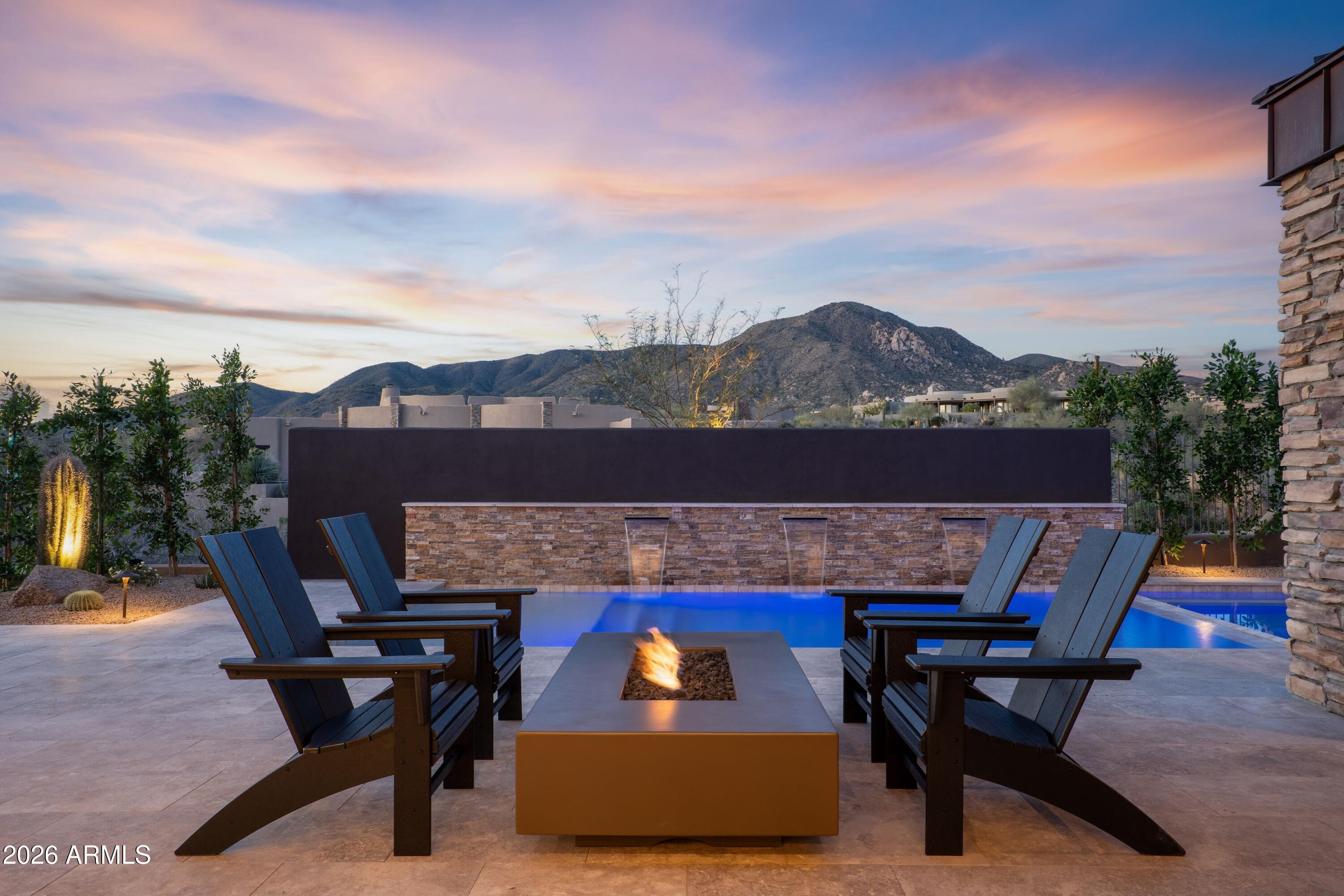 Transformed home by Aesthetic Interiors & Design! A striking fusion of casual sophistication and modern swank, this reimagined residence delivers a lifestyle defined by seamless indoor-outdoor living and elevated comfort. Every space has been intentionally crafted to feel both welcoming and artful, intimate yet bold--a home that easily transitions from quiet evenings to dazzling gatherings. The contemporary exterior sets the tone with smooth stucco, stacked-stone columns, crisp architectural lines, and a spacious modern paver motor court. Layered, Zen-inspired desert landscaping softens the approach, guiding guests through a gated courtyard where spaced concrete pads lead to a firepit lounge and the dramatic metal-accented covered entry. The mood is set immediately by a Sonos soundscape that carries throughout the home and both in front courtyard and back. The 10' glass door opens to a great room that commands attention with its floor-to-ceiling tiled fireplace and sweeping wall of windows framing majestic northern mountain vistas. The adjacent dining area glows beneath a swanky chandelier, offering a chic backdrop for intimate dinners or lively celebrations. To the left, the kitchen stands as a showpiece of contemporary design a 10' island with dual-sided bar seating, under-lit slab cabinetry, stone counters, Wolf, Subzero and Cove appliances, and a casual dining nook with built-in buffet. A 24-level, side-lit, wall-to-wall wine display bridges the kitchen and formal dining room, delivering a mesmerizing visual centerpiece worthy of a private gallery. Nearby, the den provides a luxe retreat with its black-pebble accent wall, linear fireplace, mounted TV, inset black-wood plank ceiling, and panoramic windows perfect for unwinding or cozy movie nights. The east-wing primary suite offers a haven of serenity and style. A wall of glass opens to the patio and mountain scenery, complemented by a refined dry bar. The spa-inspired bath features double vanities, a grand wet room with two walls of dual shower heads and freestanding tub, and an expansive walk-in designer closet. A nearby double-door suite ideal as an office or guest retreat echoes the home's high-end finishes with floor-level showers, eye-catching golden brass fixtures, under-lit cabinetry, and statement lighting. Two additional ensuite bedrooms on the opposite side of the home continue the theme of subtle luxury, each thoughtfully designed with the same meticulous attention to detail and a touch of swank. The outdoor living experience is extraordinary an orchestrated blend of relaxation and entertainment. Stone patios, sculpted desert landscape, and an artificial turf area surround the single-level pool and spa, enhanced by three cascading waterfalls. Multiple lounging zones, shaded dining spaces, and a raised firepit with seating invite day-to-night enjoyment. The outdoor kitchen with bar seating sets the stage for effortless alfresco dining, while view fencing ensures unbroken vistas of mountains, sunsets, and starlit skies. Located in the Village of Arrowhead, the home offers prime access to Desert Mountain's exceptional amenities: seven luxury clubhouses, seven world-class golf courses, the renowned Sonoran Fitness Center and Spa, and The Ranch a gateway to 24 miles of breathtaking trails bordering the Tonto National Forest. This mid-mountain setting delivers the perfect balance of privacy and convenience, with Scottsdale's cultural offerings, upscale shopping, and Phoenix Sky Harbor International Airport all within easy reach. More than a residence, this is a statement of living exceptionally where architectural artistry, natural majesty, and refined luxury come together in one of Arizona's most coveted communities.
