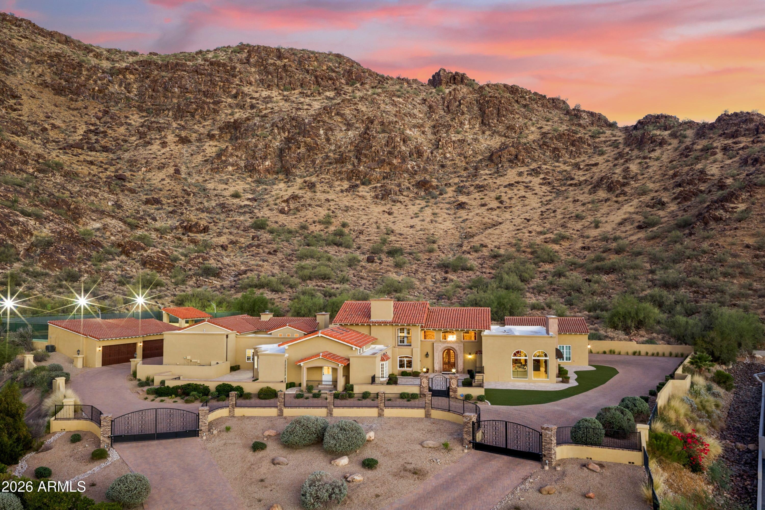 Rarely does a property of this scale, craftsmanship, and setting come to market in Paradise Valley. Nestled at the base of Mummy Mountain, this extraordinary gated luxury estate spans 9,336 square feet across four distinct living areas -- delivering the privacy of a true desert retreat with the amenities of a private resort. The main house opens through a carved stone entry arch into a two-story foyer featuring a sweeping iron-and-marble staircase, polished marble floors with a custom inlay medallion, and a crystal chandelier. Principal rooms include a grand living room anchored by a hand-carved antique fireplace surround with bas-relief medallion, a dramatic crimson-walled formal dining room, a family room with custom marble-and-hardwood checkerboard floors, and a chef's kitchen with professional 6-burner range, arched stone hood, diamond mosaic backsplash, double wall ovens, and an oversized walnut island -- flowing to a breakfast nook with brick fireplace and direct pool access. A walnut-paneled library with fireplace, formal powder room with gold mosaic vanity, and butler's pantry complete the main level. The Primary Suite occupies its own private wing with coffered tray ceiling, mood-setting rope lights, gas fireplace, and French doors to the pool terrace. The primary bath features a soaking tub beneath a vaulted arch alcove with canyon views, bookmatched Calacatta marble shower, and dual vanities. The Second Floor Residence is the original primary suite -- approximately 1,500 square feet with separate entrance, private covered patio, and spectacular mountain views. The Guest Wing offers three bedrooms -- one en-suite and two sharing a beautiful Jack and Jill bath -- with soaring vaulted ceilings throughout. The Fully Equipped Casita provides a complete independent residence with bedroom, full kitchen with Viking range, jetted tub bath, and private laundry. Outside: resort pool with fountains and spa, vast travertine terrace, circular fire pit, covered loggia with fire table, and a regulation lighted tennis court with shaded pavilion -- all framed by Mummy Mountain.