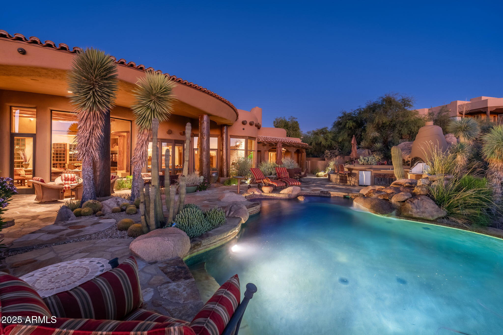 Set within the gated enclave of Pinnacle Canyon at Troon North, this extraordinary one-of-a-kind estate rests along the 16th green of the Monument Course back nine, seamlessly embedded into the Sonoran Desert landscape. The property comes fully furnished, w/ accessories & minimal exclusions, included in the asking price. Built by renowned RS Homes & masterfully designed by Urban Design Associates with interiors by Bess Jones Interiors, the residence earned national recognition, including the 2003 Award of Merit for Best in the West Excellence and the Best in American Living Award for Best One-of-a-Kind Custom Home. The home is a modern interpretation of a Native American dwelling, an organic Pueblo-inspired masterpiece where architecture, artistry, and environment exist in perfect harmon From the moment you enter, the home's sculptural form and hand-crafted detail are unmistakable. Smooth, deep sand-colored stucco, rounded edges, curved walls, kiva fireplaces, and adobe, stone, and wood accents create a timeless Southwestern aesthetic that feels both grounded and refined. Flagstone flooring flows through the main living areas, while reclaimed oak wood flooring and wood ceiling details add warmth and authenticity. Soaring ceilings and wood casement doors and windows frame dramatic views of boulders, desert vegetation, and the golf course beyond, flooding the interior with natural light.
The living and dining rooms are joined in an elegant great room designed for both daily living and entertaining. Large picture windows capture the surrounding desert and fairway views, while a built-in wood bar with sink and display cabinetry anchors the space. The dining room features a built-in wine refrigerator and a softly illuminated cove ceiling. Nearby, the powder room stands as a true work of art with a floating stone vanity, hand-finished plaster walls, custom mirror, and distinctive light fixtures.
The kitchen is nothing short of spectacular, functioning as both a culinary workspace and gathering place. Highlights include a tabletop fireplace, gas cooktop with pot filler, custom metal art backsplash, two Dacor wall ovens, and separate built-in refrigerator and freezer concealed behind hand-carved wood panels inspired by Native motifs. A center island with prep sink, reclaimed oak flooring, built-in microwave, and seamless connection to the family room make the space both functional and visually stunning. The adjacent family room features a centerpiece adobe brick fireplace and accent wall, custom wood paneling that conceals the television, and a herringbone wood ceiling with exposed beams, opening to a breakfast room and exterior living spaces. A Crestron system manages lighting, climate, and entertainment throughout the home.
The primary suite is a serene retreat featuring a private patio, separate his and her closets, dual vanities, two toilet rooms, a large soaking tub with etched glass windows for privacy, and a dramatic wrap-around shower with dual entrances. The thoughtfully designed floor plan includes a private guest wing with two spacious bedrooms, each with ensuite baths and walk-in closets, one with its own private patio, along with a dedicated guest laundry room and custom coffee cabinet with built-in refrigerator. The main laundry room is equally impressive, offering hand-painted wallpaper, sink, center island, and abundant cabinetry. In addition, a detached casita offers exceptional flexibility with its own kitchenette, private patio, and guest accommodations, perfect for visiting guests.
A standout feature of the home is the upstairs executive office, accessed via interior and exterior curved stairs, designed as a private sanctuary. This space includes a fireplace, half bath, ample storage, reclaimed oak flooring, wood ceiling accents, and a private patio, ideal for working in peace while enjoying elevated desert and golf course views. 
The exterior living experience is equally exceptional, designed to mirror the home's interior craftsmanship. Flagstone hardscape, matching the interior flooring, winds through covered patios and lounging decks. The backyard features a resort-style pool with boulder waterfall, a separate heated spa, multiple fireplaces, lush desert landscaping, and an outdoor kitchen complete with barbecue, cooktop, and sink. A three-car garage with EV charger, epoxy flooring and storage rooms completes this remarkable property. With unmatched attention to detail, architectural integrity, and award-winning design, this estate stands as a rare and iconic expression of luxury Southwest living in Troon North.