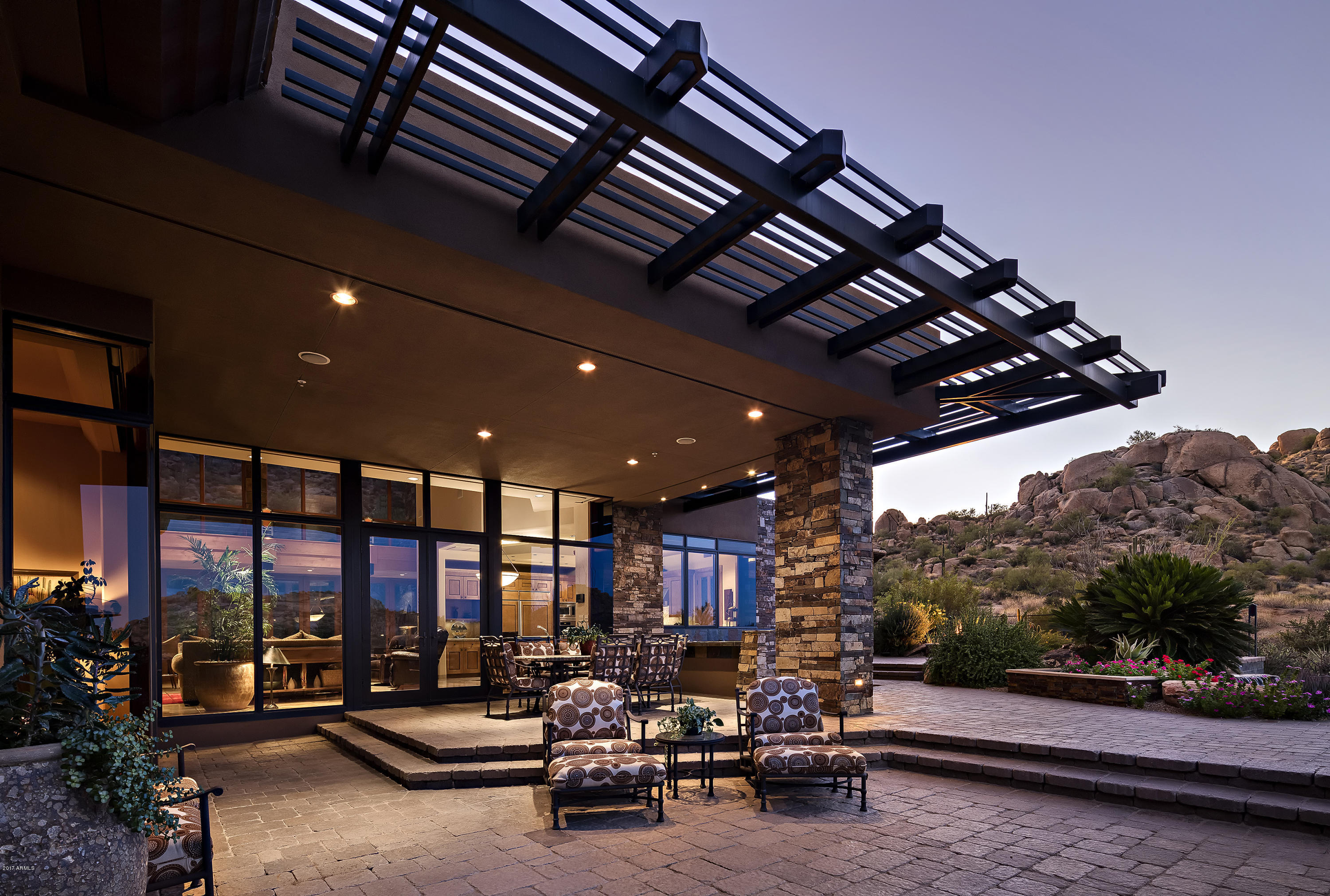 Elegant Frank Lloyd Wright inspired architecture with elevated views in the renowned community of Estancia.
Timeless finishes and impeccable details are featured in this contemporary design by Mark Sever. Materials include the use of linear metals, large glass elements, dry stack stone walls, beamed ceilings and limestone flooring. A functional and open great room floor plan with seamless indoor/outdoor living that takes advantage of the panoramic mountain and golf course views. Master suite is located on the main level with two guest suites upstairs and an attached guest house downstairs.  In addition there are his and her offices plus an exercise room. The oversized outdoor living space in the backyard includes a large covered dining patio, sundeck, an outdoor kitchen, fire pit and a heated pool and spa with water feature. The front yard living space provides a cozy sitting area tucked under Pinnacle Peak and the surrounding boulders. Sited on over 1-acre of land, this home is a masterful expression of livable and functional contemporary design.
