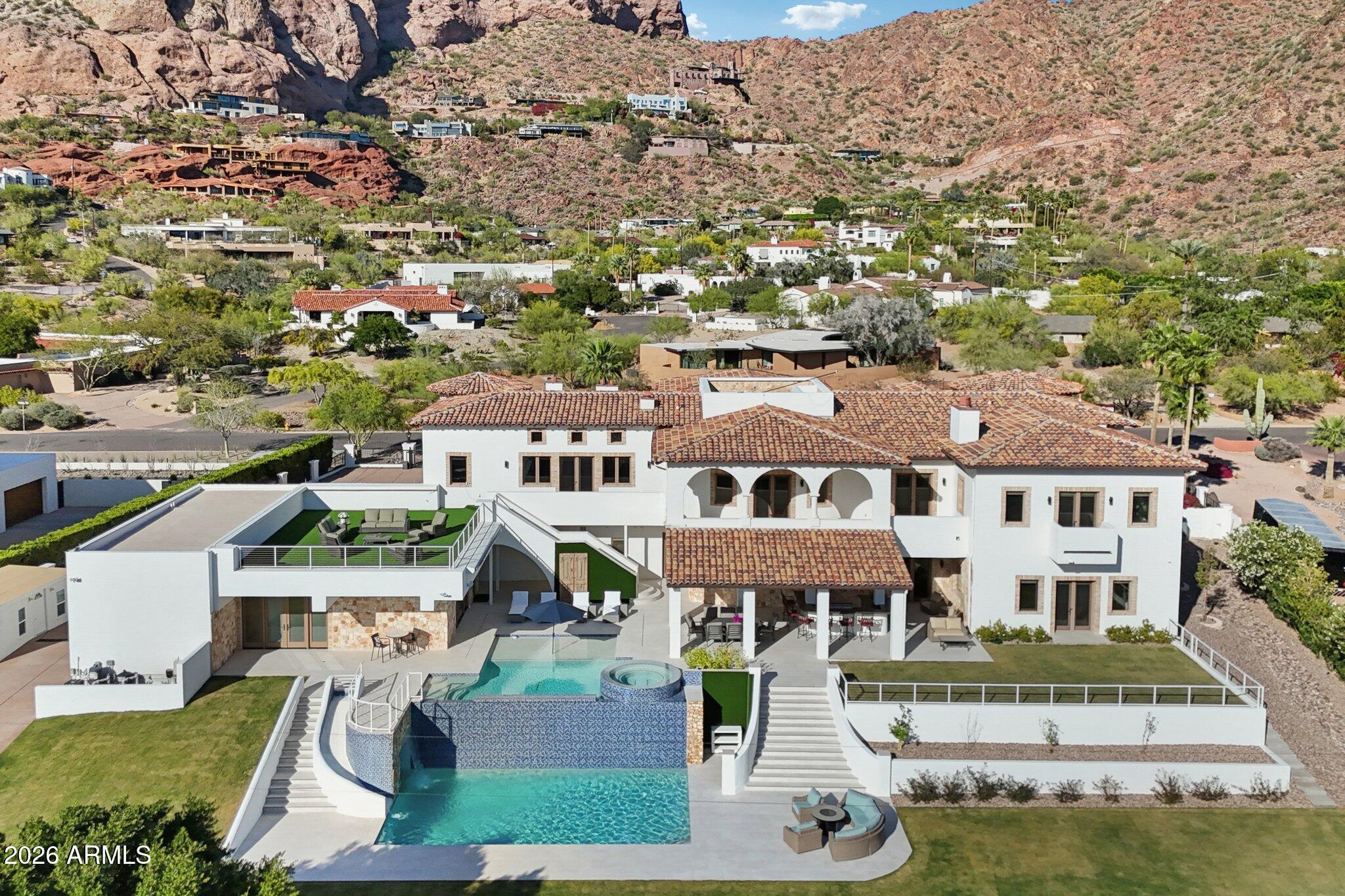 Privately gated on over 1.09 acres in prestigious Arcadia Proper -one of Phoenix's most sought after neighborhoods. The Rockridge Estate captures panoramic views of the city skyline and Camelback Mountain as its backdrop. A grand porte-cochère arrival leads to soaring, architecturally rich interiors, where a dramatic 15' wall of glass frames the view. The resort-style backyard is anchored by a two-level infinity-edge pool with a 10' water wall, grotto, and water slide. Spanning over 13,000 sqft, the estate offers 7 bedrooms, including two primary suites, 9 bathrooms, 5 fireplaces, an elevator, and expansive living spaces designed for elevated living and entertaining. The chef's kitchen serves as the heart of the home, appointed with Wolf and Sub-Zero appliances , dual cooking surfaces, and a curved run of custom cabinetry and quartzite countertops beneath a wall of windows. An expansive multi-level island, farmhouse sinks, and walk-in pantry are complemented by a secondary pantry, laundry, mudroom features, and dedicated dog spa.
The upper level, accessed from the beautiful wrought-iron railed grand staircase, or private elevator, offers a loft overlooking the main living areas and a north-facing veranda capturing Camelback views. A game room with wet bar, fireplace, en-suite bedrooms, and optional private access creates a flexible secondary living environment.
The upper-level primary suite spans approximately 2,000 sqft with fireplace, terrace, and sweeping views, complemented by a spa-inspired bath, boutique-style closet with laundry, and adjoining flex space. A rotunda office or studio and lower-level vault with wine cellar or simulator potential add further versatility.
Lush, park-like grounds unfold across multiple levels with illuminated staircases and expansive terraces. A striking two-level infinity-edge pool cascades over a 10-foot water wall, with a lower-level grotto, water slide, and lounge areas, and an upper level featuring an infinity-edge spa spilling into both tiers, in-water seating, and sun shelf. A covered patio with BBQ and bar seating for 12, motorized shades, and custom awning supports year-round enjoyment. An elevated deck and rooftop lounge capture panoramic city, mountain, and 360-degree views -creating a seamless indoor-outdoor experience at every level.
An attached two-car garage is complimented by a detached RV garage with additional bays, accommodating 10+ vehicles, recreational storage, or car collections. A dedicated RV gate and expanded parking areas maintain the integrity and flow of the grand circular drive.
Set at the base of Camelback Mountain in Arcadia Proper, with close proximity to premier dining, shopping, and lifestyle amenities.