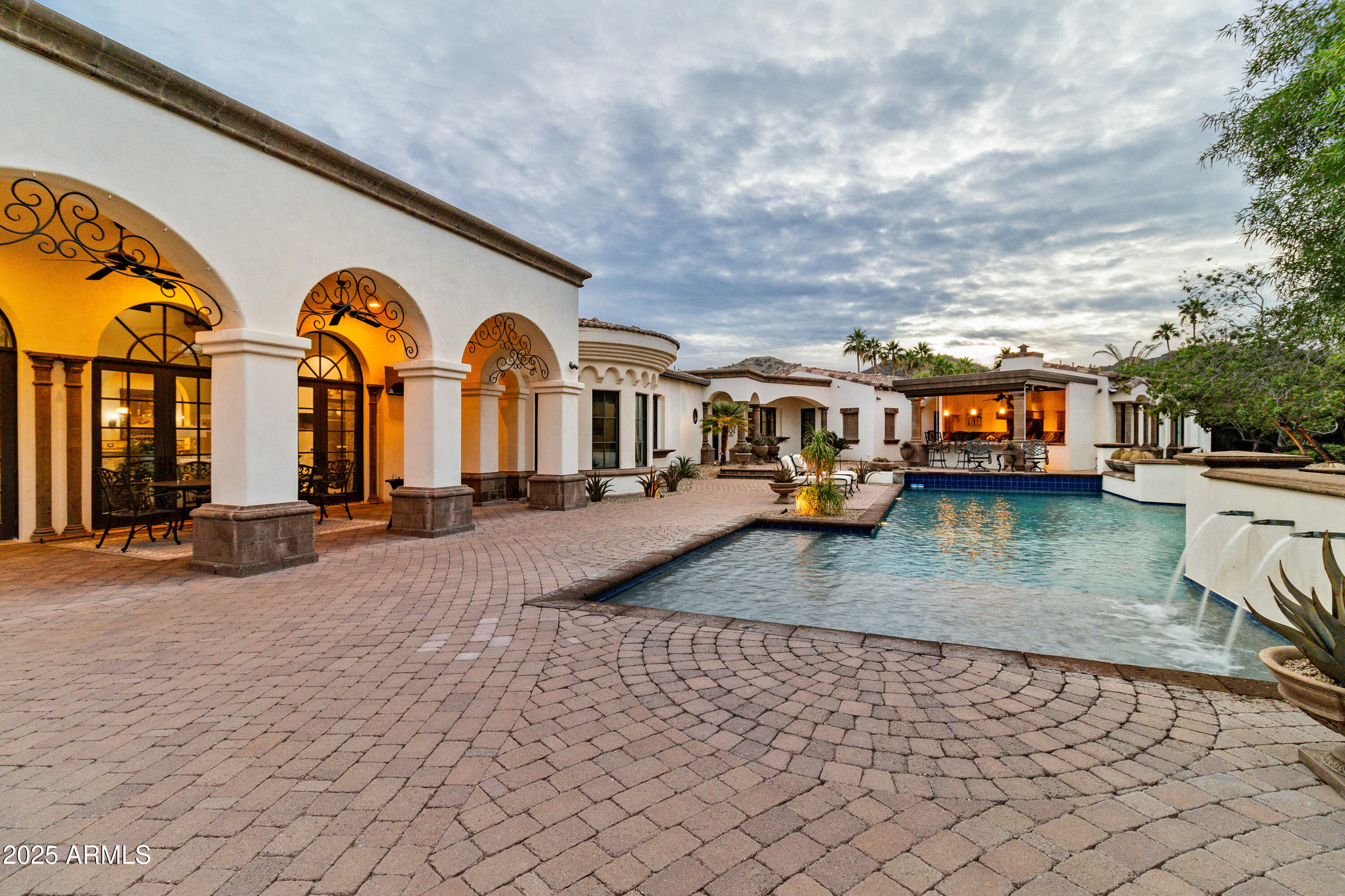 Welcome to this exquisite single-level, Santa Barbara-inspired estate nestled on a private gated lot at 6770 E Bluebird Ln in coveted Paradise Valley. With approximately 7,975 square feet (5 bedrooms, 5½ baths), this exceptional residence blends sophisticated finishes with seamless indoor-outdoor living. From the moment you arrive, the atmosphere is stunning: a gated courtyard introduces the home's architectural elegance and lush grounds. Inside, rich wood flooring, stone and marble accents, and extensive custom millwork throughout set the tone of refined luxury. Each of the five en-suite bedrooms offers a sanctuary of its own; the primary suite opens directly to a private terrace, creating a seamless extension of indoor luxury to outdoor serenity. A dedicated five-car garage adds both function and prestige. Ideal for entertaining  and everyday living  the outdoor grounds feature multiple covered patios, expansive lawn areas, fire-pits, fountains, and a sparkling pool and spa. For the chef at heart, the extraordinary outdoor kitchen and bar elevate this home to true resort-style living. Located in one of the Valley's most prestigious neighborhoods, this home offers the blend of privacy, luxury and sunshine that defines the Paradise Valley lifestyle. With a generous lot, upscale finishes, and premium resort-style amenities, this estate is a rare offering.
