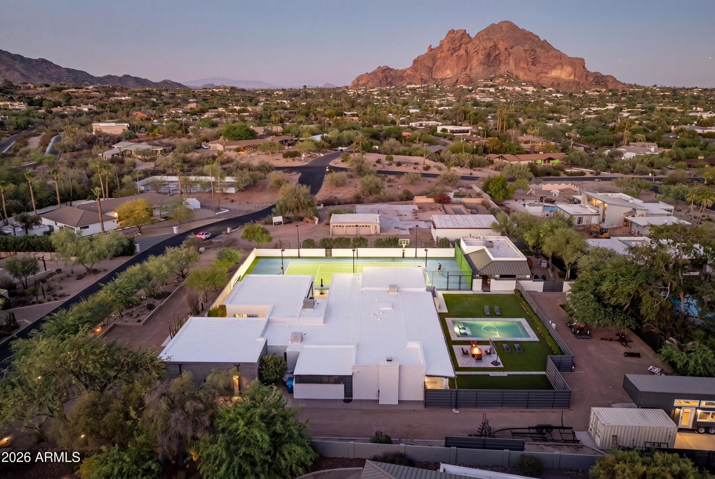 Ideally set on a full acre in the heart of the Camelback Corridor, this modern-transitional estate offers iconic Camelback Mountain views, resort-caliber grounds, and a private lifestyle that is truly special. Spanning over 4,800 sqft across a main residence and a fully independent 2-bed, 1-bath guest house, the property offers 6 bedrooms and 5.5 baths in total - a rare combination of space, privacy, and flexibility in one of Paradise Valley's most coveted corridors. Clean white stucco, dark steel-framed windows and doors, warm wood ceiling accents, and a private paver courtyard entry reflect architecture that is refined without being overdone, with a natural indoor-outdoor flow throughout. The grounds tell the full story: a lighted tennis court with basketball, a sparkling pool set within manicured turf, a fire pit seating area, and a covered outdoor kitchen and dining area with a pass-through window from the kitchen. The crown jewel is the sport court pavilion -- a fully appointed indoor-outdoor entertaining space with glass garage doors that open completely to the tennis court. Day or night, this property lives like a private resort. No HOA. Steps from Phoenix Country Day School and minutes from Brophy College Preparatory, Xavier College Preparatory, Sanctuary Camelback Mountain Resort, and the best dining and retail the Valley has to offer.