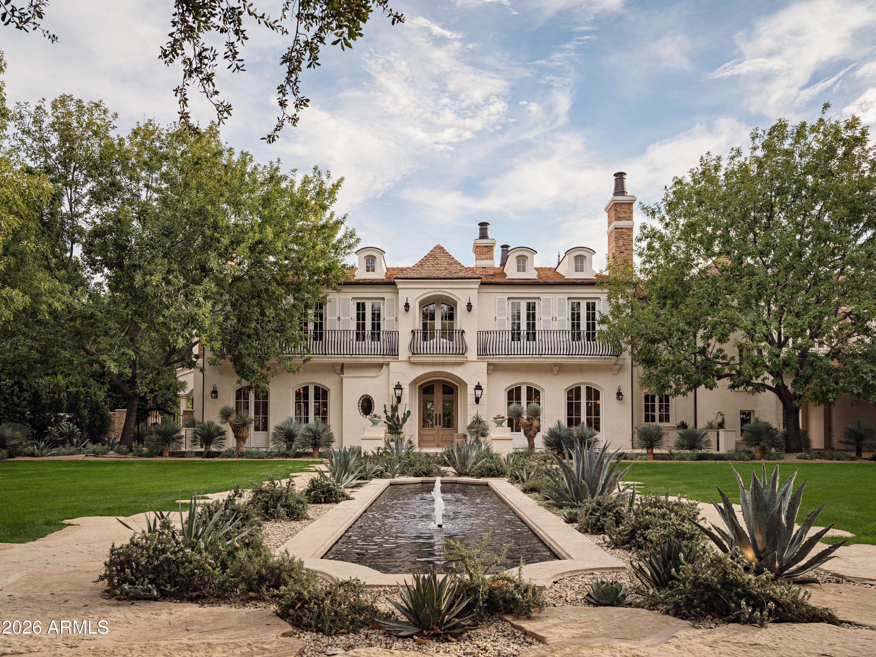 Behind tall private iron gates on the most coveted stretch of Exeter Boulevard, this extraordinary French-inspired Arcadia estate sits on nearly a full acre with commanding street presence, a dramatic fountain arrival, and rare north-south exposure—significantly updated and free of overhead power lines. Designed by architect Mark Candelaria and built by Nance Construction, the residence showcases authentic Old-World masonry construction and timeless European elegance paired with modern luxury. Interiors are defined by elegant archways, grand formal and informal living spaces, and a dramatic dual-foyer stair hall. Reclaimed wood beams, imported limestone and European oak floors, custom millwork, Niermann Weeks lighting, and carved limestone fireplaces with reclaimed firebrick reflect meticulous attention to detail. The chef's kitchen features a statement French range and professional-grade appliances, complemented by an adjacent catering pantry and full bar. Additional highlights include a library, glass conservatory, and climate-controlled wine cellar.
Grand curved staircases or an elevator lead to the primary suite, a private retreat with sitting room and fireplace, view balconies, and a boutique-caliber dressing room. The spa-inspired primary bath offers a steam shower, dual water closets, and a fireplace beside a nickel-clad soaking tub. Three additional en-suite bedroomsone ideal as a gym with steam showeralong with an upstairs lounge or study and second laundry room complete the upper level. A detached guest casita provides full living quarters with kitchen, fireplace, bedroom suite, and balcony. Resort-style grounds feature multiple covered patios with fireplaces, a soaking pool with integrated fountains, a heated pool cabana with bar and bath, pickleball and bocce courts, and lush gardens accented by reclaimed brick, cobblestone, sandstone, gas lanterns, and water featuresevoking the ambiance of a European villa.  A recent, comprehensive renovation includes new major systems, updated home automation, and extensive landscape enhancements. With temperature-controlled garage parking for up to ten vehicles, exceptional privacy, and uncompromising quality, this is a rare, legacy estate in one of Arcadia's most prestigious settings.