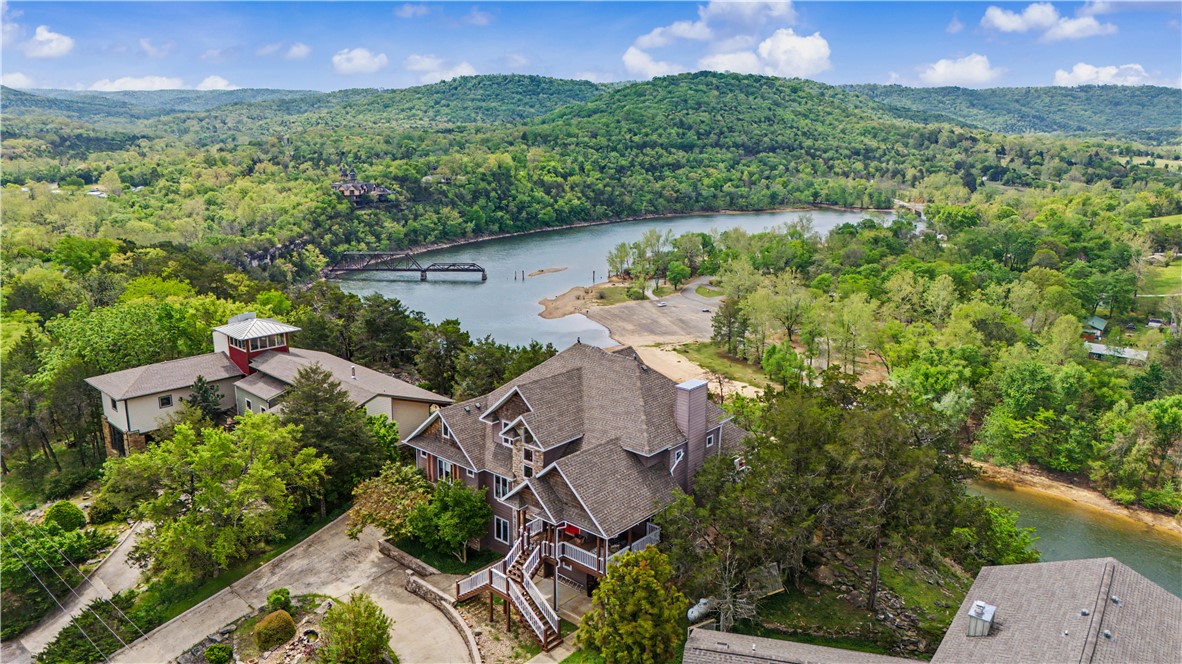 This stunning home is perched on a bluff with sweeping views of Table Rock Lake, Castle Rogue's Manor, the "Little Golden Gate Bridge" over the White River/Table Rock Lake in a historic town. Ascending the front steps, a covered porch leads to double entry doors. Inside, a grand living room boasts vaulted ceilings with a stone fireplace, wood floors and lake views. A large primary suite with deck access overlooking the limestone bluffs sits just off the kitchen and living area on the main level. The upper level offers a balcony overlooking the living room, sitting area, 2 bedrooms or bonus spaces and a bathroom. Downstairs, the basement features a bedroom, bath, living area and workshop plus garage access. The home is equipped for an elevator setup. Large deck areas maximize the home's viewing position, and terraced, stone-accented landscaping blends in subtly. Situated in the planned community of Holiday Island, this home offers access to the Table Rock Marina, recreation amenities, and a unique, vacation vibe.