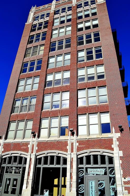 There are lofts that check the boxes, and then there are lofts that remind you why people fall in love with downtown living in the first place. This corner residence at Printers Lofts brings together the best parts of the St. Louis loft experience: historic character, natural light, modern convenience, garage parking, and a building community that actually feels like a community.

Inside, the space is defined by soaring ceilings, exposed brick, and an extraordinary wall of 14 oversized 8-foot windows that wrap the corner and fill the loft with light. Fully automated custom blinds make the space easy to live in throughout the day, whether you want bright morning energy, filtered afternoon light, or a quieter evening feel.

The open layout works beautifully for both daily living and entertaining. The kitchen is a standout, with paneled KitchenAid and Bosch stainless steel appliances, contemporary cabinetry, and a generous center island that naturally becomes the gathering place — for coffee, dinner prep, or friends stopping by before a night out downtown.

The bedroom area is thoughtfully positioned for privacy and includes generous closet space, while the modern bathroom has a clean, refined feel with smart touches like occupancy-sensing, no-touch lighting. The in-unit laundry area includes the same hands-free lighting, adding one of those small conveniences you appreciate more every time you use it.

The loft is also well equipped for how people actually live now, with a smart thermostat, keyless Yale entry, and available gigabit fiber internet for remote work, streaming, and everyday connectivity. An assigned space in the heated, secure garage is included, along with secure building entry and a transferable Hoffman Bros maintenance plan for added peace of mind.

Printers Lofts spans two sister buildings at 1611 and 1627 Locust, connected by a shared underground garage. This residence is located in 1627, home to the furnished rooftop deck with grills, comfortable seating, and wide-open sunset views over the St. Louis skyline. Residents also enjoy access to 1611’s second-floor community room and fitness center, giving the building shared amenity space that is genuinely useful, not just decorative.

What makes Printers Lofts especially compelling is the balance: historic architecture without giving up comfort, a downtown location without sacrificing parking, and an established HOA with a welcoming residential feel. From here, Washington Avenue restaurants, coffee shops, nightlife, CityPark, Enterprise Center, and the broader downtown neighborhood are all part of daily life rather than occasional destinations.

For buyers looking for a true downtown loft — not just an apartment with brick walls, but a home with light, presence, convenience, and a real sense of place — this one deserves a closer look.