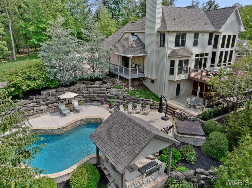Discover your private sanctuary on over 3 wooded acres—an elegant custom retreat designed for privacy, comfort, and unforgettable entertaining. Enjoy your outdoor paradise featuring a saltwater pool with swim-up bar, spa, covered outdoor kitchen, full pool bath, and laundry with private entry. Relax on the composite deck overlooking the pool, complete with stone fireplace and built-in Green Egg grill. Step inside to a dramatic 2-story great room with soaring ceilings, a wall of windows, gas fireplace, and built-ins, flowing seamlessly into the gourmet kitchen with granite counters, large island, GE Monogram gas cooktop and hood, a stone backsplash, breakfast bar, and see-through wet bar. Adjacent is the hearth room with a floor-to-ceiling stone fireplace, offering access to a covered porch, perfect for relaxing year-round. The main-floor primary suite features a coffered ceiling, dual-sided fireplace, custom walk-in closet, and a spa-inspired bath with jetted tub, vaulted ceiling, walk-in shower, and double vanities. A breakfast room, coffered-ceiling dining room, and private office with built-ins complete the main level. Upstairs includes a loft overlooking the great room, with two spacious bedrooms and two full baths. The finished walk-out lower level offers 9' ceilings, abundant natural light, rec room with gas fireplace, bedroom, full bath, game room w/ bar, safe room w/ custom cabinets, workbench, and a wine/bourbon cellar. All just minutes from Missouri wine country.