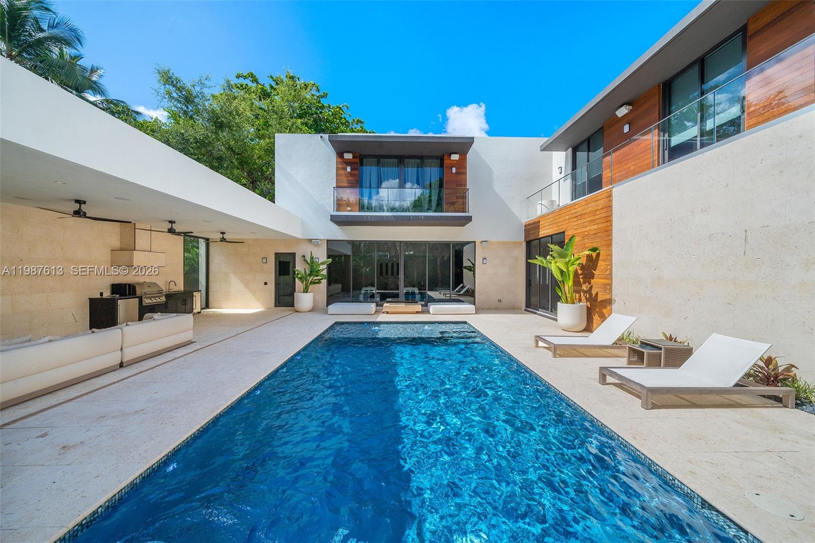 This luxurious 5-bedroom, 6-bath Florida Modern residence boasts over 5,500 square feet of elegantly designed indoor and outdoor living space, nestled among lush tropical landscaping for ultimate privacy and tranquility. Floor-to-ceiling windows bathe the interiors in natural light, highlighting an open-concept layout that effortlessly blends indoor sophistication with outdoor serenity—ideal for entertaining. A dramatic dining area with soaring 22-foot ceilings and a striking floating staircase sets the tone for the home’s impressive design. The impeccably curated Armazem kitchen—with a custom wine cellar—flows into a spacious great room that opens to a covered poolside terrace featuring a fully equipped outdoor kitchen with an icemaker.