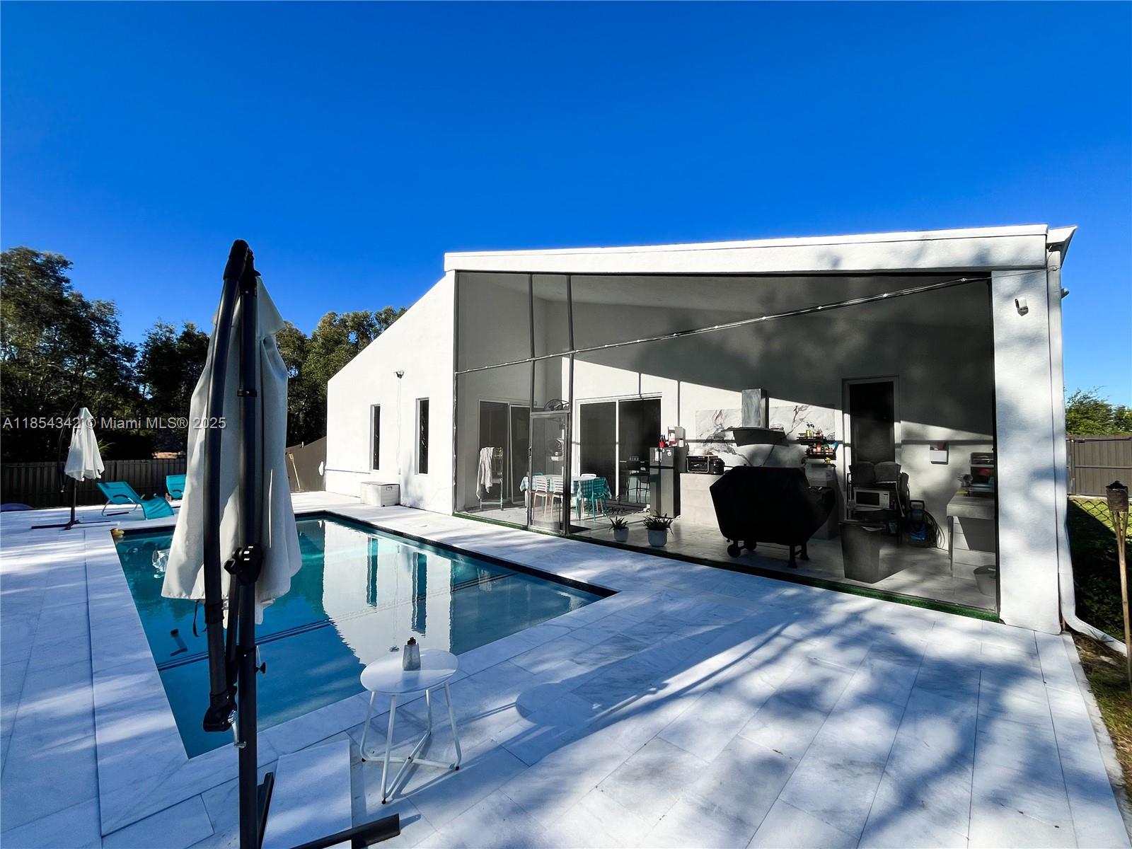 Architecturally distinctive modern home with soaring 18-ft ceilings. The only new-build in Lehigh under a half-million to offer a pool! Tall impact windows fill the open plan in natural light, while large-format tile grounds every room. Unlike typical builds, this home has full CBS construction—including interior walls—for enhanced soundproofing and superior storm protection. Furnished living room, outdoor loungers, umbrellas, electric fireplace, RO system, security cameras, appliances, washer/dryer & finished closets all convey. 3-BD/2-BA plan + private den. Screened, covered patio holds full outdoor kitchen; beyond, a 12×28 travertine-decked pool with tanning ledge invites effortless indoor-outdoor living. Oversized 2-car garage, broad drive, generous fenced lot. No HOA. Ready now.