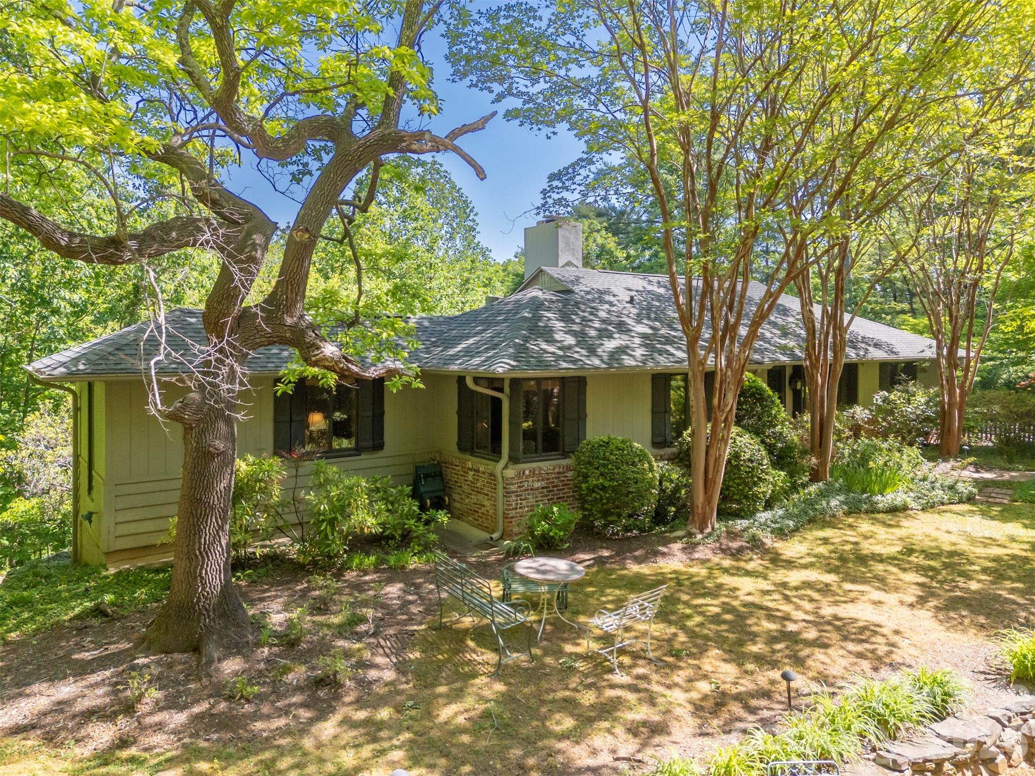 Tucked beneath majestic Warrior Mountain on 7.2 private ridge-top acres at the end of a quiet road, just minutes from downtown Tryon, this extraordinary retreat offers the rare feeling of living among the trees. Birdsongs replace traffic noise, and every window frames layered canopy views that change beautifully with the seasons. Meandering stone paths wind through a gently sloping sculpture garden, leading to hidden seating areas, lower parking, storage spaces, and even a wooded trail descending toward a running creek below.
Designed for both relaxation and entertaining, the home blends rustic luxury with artistic character. Warm wood-paneled walls, handsome beams in the dining room, built in bookshelves and three gas log fireplaces create inviting gathering spaces throughout the home — including fireplaces in the formal den, dining/family room, and primary suite. A dramatic wall of large sliding windows, craftily screened, create the perfect setting for unforgettable Carolina spring and fall dinner parties immersed in the open air amongst the treetops.
The detached two-car carport doubles as an outdoor entertaining pavilion with generous seating areas overlooking the grounds, while additional garden seating invites quiet mornings and evening conversations under the trees.
The lower level offers a walk-out apartment with kitchenette and eclectic, artistic bath arrangements — ideal for guests, extended family, studio space, or creative retreat possibilities.
Private, peaceful, and unlike anything else on the market, this one-of-a-kind mountain sanctuary captures the essence of refined woodland living in the heart of the Carolina foothills.