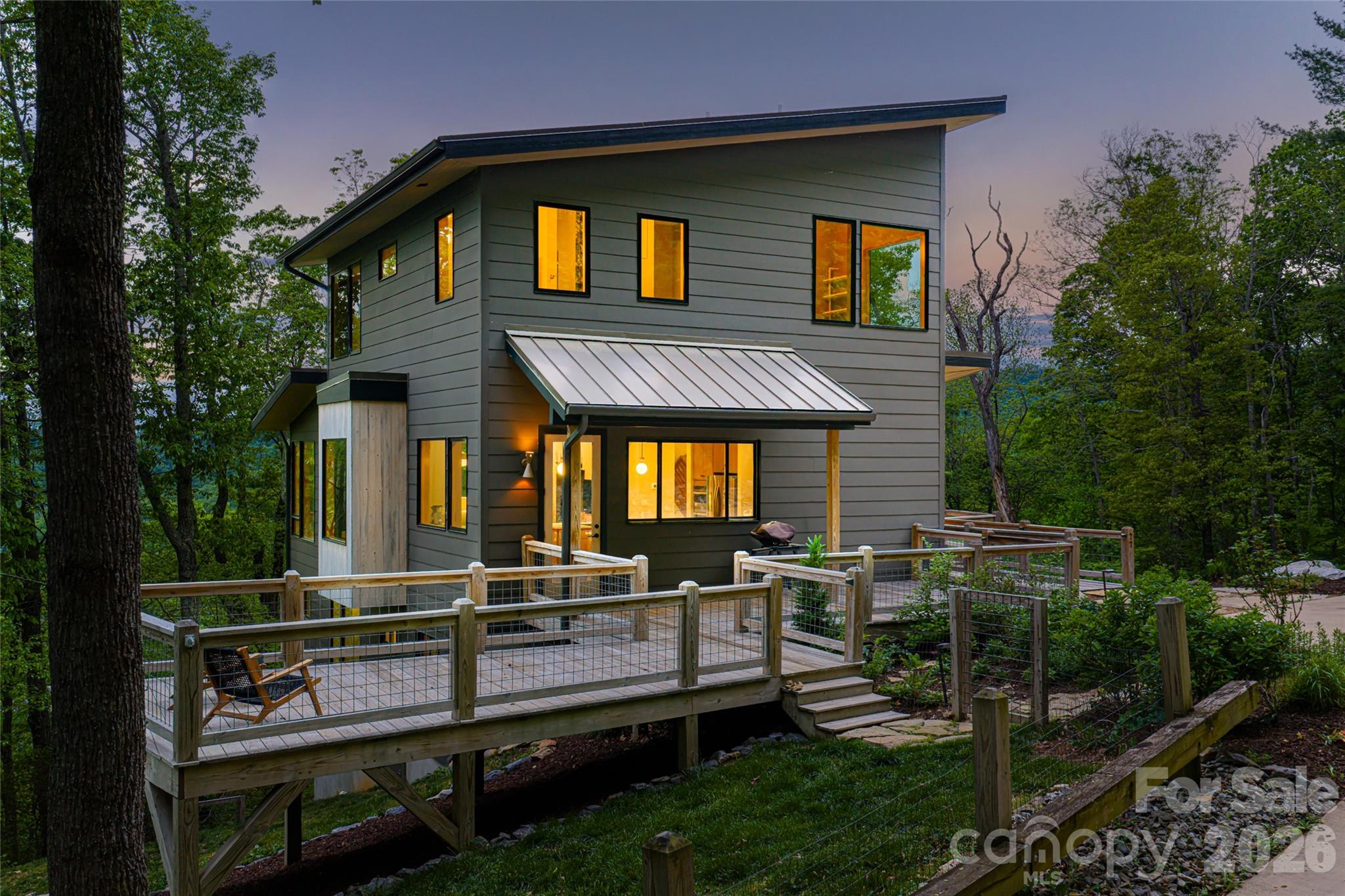 At the end of a quiet cul-de-sac in the Glen Merrill neighborhood of South Asheville, this 19-acre modern mountain retreat is a rare expression of privacy, light and landscape. A long, winding approach sets the tone, offering a sense of arrival that feels serene and removed, while unveiling breathtaking, layered mountain views that define the home’s setting. Designed to embrace its natural surroundings, expansive decks extend the living spaces outdoors, inviting morning coffee at sunrise and evenings framed by ever-changing skies. Inside, a thoughtfully designed drop zone provides everyday convenience, while the main level unfolds into an open-concept living space that is refined and welcoming. The kitchen, dining area and living room flow seamlessly together, anchored by warm wood floors and surrounded by generous windows that flood the home with natural light and bring in the outdoors. Upstairs, the private primary suite offers a peaceful sanctuary, complemented by a versatile bonus room ideal for a studio, lounge or additional workspace. The lower level expands the home’s livability with two bedrooms, two baths, a recreation area, dedicated office and a well-appointed laundry room, ideally for guests or multi-generational living. A detached one-car garage adds to the property’s functionality, while the surrounding landscape ensures a sense of quiet seclusion rarely found so close to Asheville’s vibrant offerings. This is a home where modern design meets mountain tranquility, crafted for those who value connection and retreat. Extensive private trail system throughout the acreage in this incredibly convenient location in South Asheville. Home completed in 2021.
