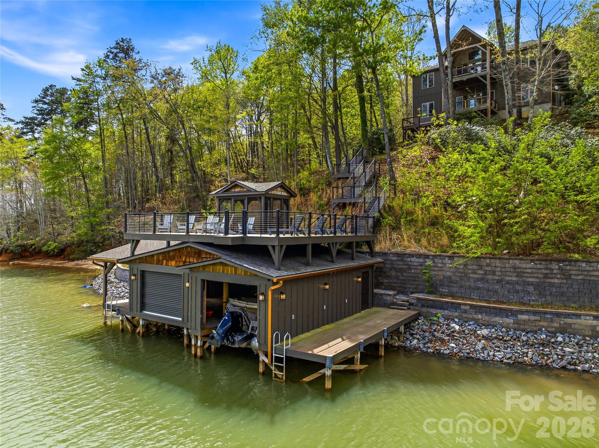 Builder’s custom lakefront double lot retreat, showcasing mountain elegance in a private setting. Located on a deep water main channel in the Gated Community of Blue Heron Point on Lake Lure. Privacy abounds on this rare double lot with over 200 feet of shoreline. This property features a private beach, and an exceptional new boathouse with party deck above. Inside, the home is rich with warm wood finishes and thoughtful details, including post-and-timber exposed beams, tongue-and-groove cathedral ceilings, granite countertops, stainless steel appliances, and a striking stone fireplace. The split-bedroom floor plan enhances privacy, while double covered lakeside decks provide stunning views from multiple vantage points. The main level of the home offers an open-concept design connecting the living, dining, and kitchen areas—perfect for gathering and entertaining. The spacious primary suite, complete with a large private bath and walk-in closet, is thoughtfully positioned for privacy, while two guest bedrooms with a Jack-and-Jill bath are located on the opposite side of the home. The lower level expands your living space with a generous family room, full bath, bunk room, and game room, all opening to the lakeside mountain views and deck. Two additional unfinished rooms currently used for game room and storage provide excellent potential for expansion—ideal for a home office or additional bedrooms. Step outside to enjoy true lakefront living. The lower deck leads directly to the hot tub area then down to the boathouse and waterfront, where you can swim, fish, or relax on your private beach. The boathouse is among the finest on Lake Lure, featuring double slips with barn-door access, covered outdoor living space, side docks for paddleboards, and a screened upper-level porch perfect for lounging or entertaining. This very private property offers the perfect blend of luxury, comfort, and natural beauty—an ideal full-time residence or mountain lake retreat on the main channel of Lake Lure.
