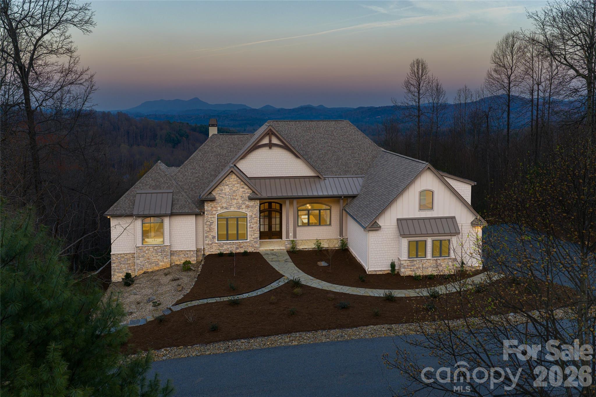 Set within the mountain enclave of Cobblestone Village, 527 Falkirk Way offers a rare opportunity to own a new custom residence with expansive, long-range mountain views. A flagstone walkway, Hardie-shake siding and stone accents introduce a home defined by thoughtful craftsmanship and scale. The main level features oak hardwood floors and a light-filled, open-concept design anchored by a vaulted great room with exposed beams, gas fireplace and custom built-ins, all oriented toward the Blue Ridge backdrop. The kitchen blends form and function with granite countertops, a large island, walk-in pantry, and a curated appliance suite including a Fulgor Milano 36-inch range and wall oven. A built-in banquette adds warmth, while living spaces extend seamlessly to a covered deck with vaulted ceilings and a stone fireplace for year-round enjoyment. The primary suite offers a private retreat with coffered ceiling, sitting area, dual walk-in closets and direct deck access. The en-suite bath features heated tile floors, dual vanities, a soaking tub and walk-in shower. An executive office, well-appointed laundry room and drop zone from the three-car garage enhance everyday livability. An elevator connects to the lower level, where a spacious recreation room with gas fireplace and wet bar opens to a covered patio. Additional bedrooms and flexible living spaces. including a steam shower, provide comfort and versatility for guests. Above the garage, a private en suite offers ideal flexibility for a studio, office or guest suite. Modern systems include a zoned Trane gas furnace with humidifier, tankless water heater, central vacuum and whole-home Generac generator. On over three acres, the property offers privacy and room to expand outdoor living, including a fire pit area. Minutes from Flat Rock, Hendersonville, and with easy access to Asheville and Greenville, this turnkey mountain residence pairs new construction quality with timeless design and exceptional views.