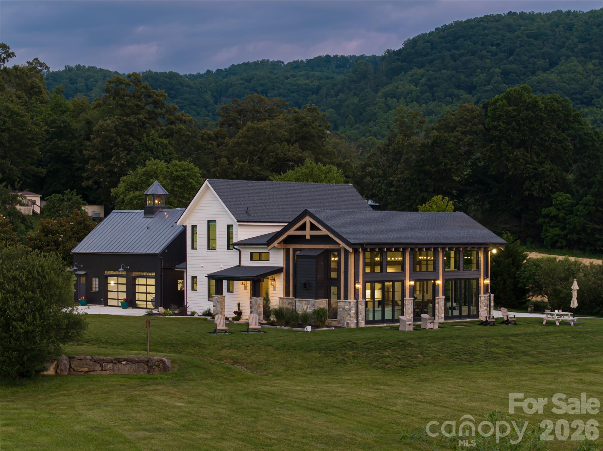 This dreamy newer-build farm estate in Cane Creek offers a blend of privacy, timeless design and natural beauty. Introduced by a sweeping driveway winding beneath a canopy of mature trees, the approach sets a storybook tone—one that feels lifted from the pages of a country magazine. Meadow-like acreage, a tranquil pond, and lush foliage surround the home, creating a private sanctuary where serenity and sophistication coexist. Constructed in phases—the garage wing in 2013 and the main house addition in 2023—the residence reflects enduring utility and fresh architectural vision. Clad in rustic wood, steel and exposed timber, the exterior embodies the warmth of classic farmhouse style while embracing the clean lines of contemporary design. Inside, meticulous craftsmanship and natural textures continue the narrative. Vaulted ceilings and abundant windows flood interiors with light, framing pastoral views and drawing the outdoors in. The heart of the home is its open-concept living, dining and kitchen spaces—designed for gatherings that are intimate and grand. Rich woodwork and exposed beams create a sense of history, while modern finishes offer comfort and convenience. The kitchen blends rustic charm with function, anchored by wide counters and premium appliances, with thoughtful touches like a walk-in pantry and dedicated coffee bar. Throughout, every detail reflects a balance of artistry and livability. Life here unfolds with ease. Mornings begin with coffee overlooking the pond, afternoons invite gardening, exploration or quiet walks beneath the trees, and evenings conclude with fireflies glowing across the meadows. Practical features, including a whole-house generator, ensure resilience without compromising charm. The estate’s location—just 20 minutes from Asheville Regional Airport—offers a rare mix of seclusion and accessibility. An additional five acres are available for those seeking even greater opportunity—whether for equestrian pursuits, expanded gardens or multi-generational living. Rarely does a property so ideally capture the essence of country living while remaining so convenient to Asheville’s vibrant culture. More than a home, this is a sanctuary—an invitation to retreat, to expand and to live beautifully.