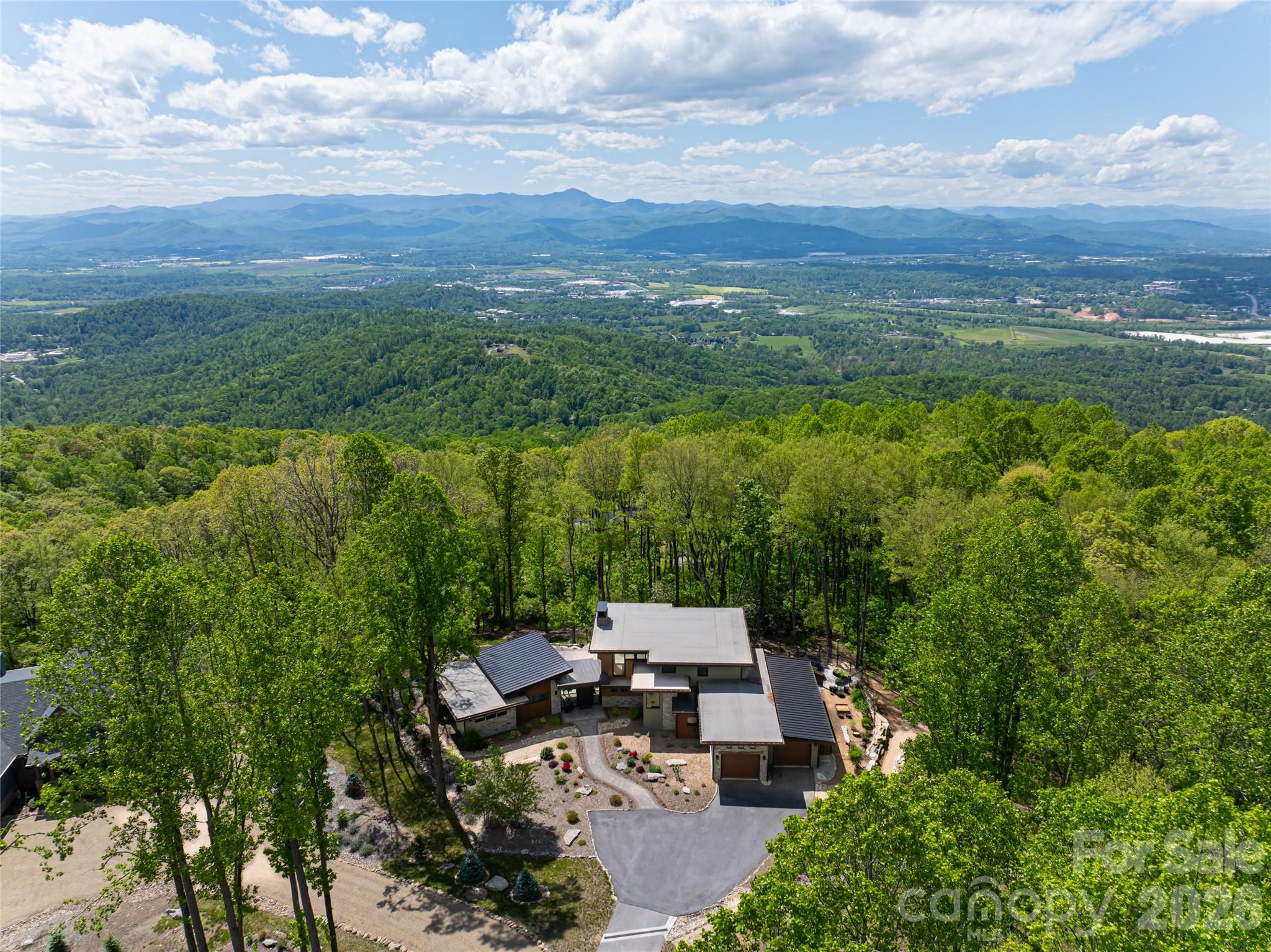 Embrace refined mountain living in this striking contemporary estate, where serenity and sophistication converge atop a rare flat mountaintop setting in the gated conservation community of Couch Mountain. Sweeping 180-degree Blue Ridge Mountain views unfold through expansive walls of glass, creating a breathtaking backdrop from nearly every room while timeless craftsmanship and elevated design define the living experience. This exceptional property is offered three separate ways to suit a variety of lifestyles and investment opportunities and this listing includes the residence and the individual lot it sits on. The home is also available with the individual lot and the adjoining lot, and the additional homesite may be purchased separately. Together, the offerings present a rare opportunity to secure privacy, flexibility and long-term value in one of Western North Carolina’s distinctive mountain communities. Inside, the dramatic great room showcases soaring 20-foot ceilings, hand-hewn 125-year-old beams reclaimed from a Canadian barn, exposed custom steel supports, and a stunning stacked-stone fireplace anchored by a concrete hearth. Designed for grand entertaining and comfortable everyday living, the open-concept floor plan flows effortlessly into the gourmet kitchen, where Thermador stainless steel appliances, Italian quartzite countertops, and bespoke rustic alder cabinetry create a space that is as functional as it is beautiful. A walk-in pantry and coffee bar, concealed behind a sliding knotty alder barn door, add warmth and character. The adjoining dining area and sunroom, with its inviting stone fireplace and panoramic mountain vistas, offer seamless indoor-outdoor living and memorable gathering spaces year-round. The main-level primary suite is a luxurious retreat featuring breathtaking views, a spa-inspired oversized shower, and a custom walk-in closet thoughtfully designed for comfort and organization. Upstairs, two private en-suite guest bedrooms showcase artisanal tilework and refined finishes, providing elevated accommodations for family and visitors alike. Outdoors, the curated mountain landscape invite exploration and relaxation with established gardens, walking trails, natural stone features and a fenced vegetable garden. Whether enjoying sunrise coffee on the terrace, entertaining beneath the stars or simply taking in the stillness of the mountains, the setting is peaceful and inspiring. Ideally just 15 minutes from Asheville Regional Airport and conveniently between Asheville and Hendersonville, the property offers easy access to acclaimed restaurants, shopping, arts and outdoor recreation. Community-approved seven-day rentals also create the opportunity for a lock-and-leave lifestyle with potential income-producing flexibility when not in residence. Rarely does a property combine architectural distinction, panoramic views, privacy and versatility so seamlessly in one extraordinary mountain offering.