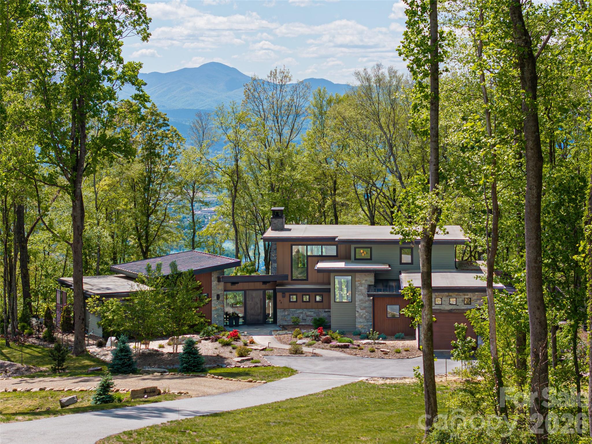 Embrace refined mountain living in this striking contemporary estate, where serenity and sophistication converge atop a rare flat mountaintop setting in the gated conservation community of Couch Mountain. Sweeping 180-degree Blue Ridge Mountain views unfold through expansive walls of glass, creating a breathtaking backdrop from nearly every room while timeless craftsmanship and elevated design define the living experience. This exceptional property is being offered three separate ways to suit a variety of lifestyles and investment opportunities as this listing includes the residence and the lot it is on plus the adjoining lot. The home is also available with only the individual lot, or the additional homesite may be purchased separately. Together, the offerings present a rare opportunity to secure privacy, flexibility and long-term value in one of Western North Carolina’s distinctive mountain communities. Inside, the dramatic great room showcases soaring 20-foot ceilings, hand-hewn 125-year-old beams reclaimed from a Canadian barn, exposed custom steel supports and a stunning stacked-stone fireplace anchored by a concrete hearth. Designed for grand entertaining and comfortable everyday living, the open-concept floor plan flows effortlessly into the gourmet kitchen, where Thermador stainless steel appliances, Italian quartzite countertops and bespoke rustic alder cabinetry create a space that is as functional as it is beautiful. A walk-in pantry and coffee bar, concealed behind a sliding knotty alder barn door, add warmth and character. The adjoining dining area and sunroom, with its inviting stone fireplace and panoramic mountain vistas, offer seamless indoor-outdoor living and memorable gathering spaces year-round. The main-level primary suite is a luxurious retreat featuring breathtaking views, a spa-inspired oversized shower, and a custom walk-in closet thoughtfully designed for comfort and organization. Upstairs, two private en-suite guest bedrooms showcase artisanal tilework and refined finishes, providing elevated accommodations for family and visitors alike. Outdoors, the curated mountain landscape invite exploration and relaxation with established gardens, walking trails, natural stone features and a fenced vegetable garden. Whether enjoying sunrise coffee on the terrace, entertaining beneath the stars or simply taking in the stillness of the mountains, the setting is peaceful and inspiring. Ideally just 15 minutes from Asheville Regional Airport and conveniently between Asheville and Hendersonville, the property offers easy access to acclaimed restaurants, shopping, arts and outdoor recreation. Community-approved seven-day rentals also create opportunity for a lock-and-leave lifestyle with potential income-producing flexibility when not in residence. Rarely does a property combine architectural distinction, panoramic views, privacy and versatility so seamlessly in one extraordinary mountain offering.