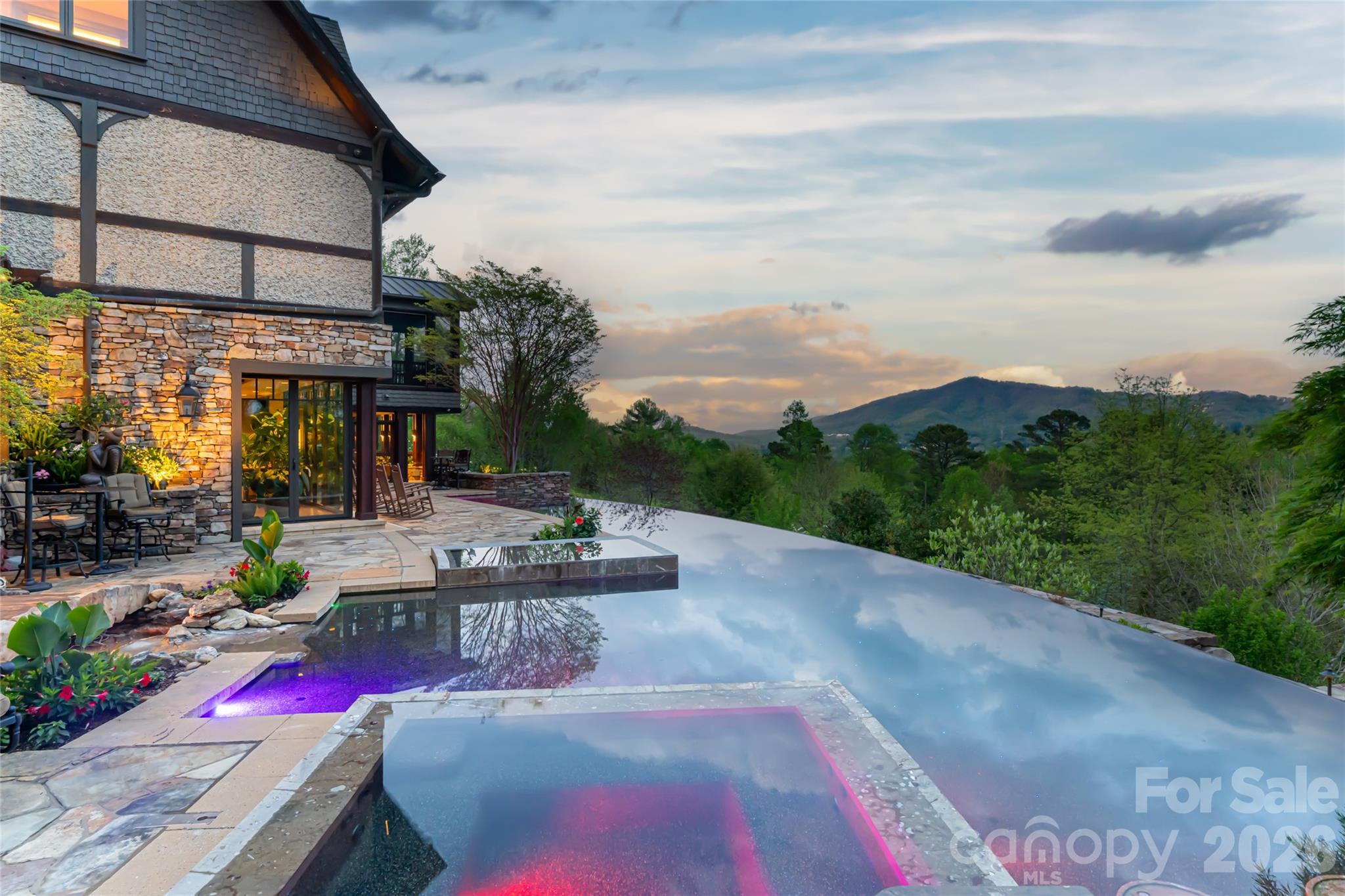 Just minutes from downtown Asheville, 66 North Deerhaven Lane is an extraordinary gated luxury estate in Asheville, N.C. offering over five acres of private gardens, award-winning landscape design and refined mountain living in the heart of the Blue Ridge Mountains. Known as Deerhaven Gardens, this remarkable property combines architectural elegance, world-class outdoor living and exceptional privacy just minutes from Asheville’s dining, arts and cultural amenities. The residences were completely renovated and expanded in 2011 with the estate’s grounds were masterfully designed by renowned Asheville landscape architect Charles Clay Mooney, whose work at Deerhaven Gardens earned the American Society of Landscape Architects’ highest national award. The property is centered around a spectacular 77-foot infinity-edge swimming pool with cascading waterfalls, natural stone terraces, streams and botanical gardens, recognized by Luxury Pools & Outdoor Living Magazine as Best Overall Outdoor Living Design in the United States. The main residence is designed to seamlessly blend indoor and outdoor living. A dramatic great room with vaulted ceilings, exposed beams and tongue-and-groove paneling opens through more than 200 feet of NanaWall accordion glass doors, creating an indoor-outdoor mountain retreat. Imported stone surfaces and natural materials complement the surrounding landscape while providing sophisticated luxury finishes throughout. At center of the home, the chef’s kitchen features a 60-inch Wolf range, Sub-Zero refrigeration, dual Miele dishwashers and custom cabinetry, opening to expansive living and dining spaces ideal for entertaining. Modern systems include fully integrated smart-home technology, geothermal HVAC, backup generators and advanced lighting, security and climate controls. The primary suite offers a private sanctuary with fireplace, spa-inspired bath, heated floors, steam shower, jetted soaking tub and custom cabinetry. Upstairs guest suites and an executive office capture sweeping sunrise views across the Blue Ridge Mountains. The terrace level recreation wing includes a media lounge, billiards room, full bar, wellness room, and fitness studio with direct access to the pool terrace and gardens. A separate guest house provides exceptional flexibility with its kitchen, living room, primary suite, bunk room and recreation space, ideal for hosting extended guests, multigenerational living or private staff accommodations. Outdoor amenities are equally impressive and include multiple entertaining terraces, a screened porch with hibachi grill, poolside summer kitchen and bar, and a private tennis court with observation deck and outdoor wet bar. Meandering paths wind through curated botanical gardens, tranquil ponds and wooded trails, creating one of the most remarkable private estates in the Southeastern United States. Despite its serene setting, the property is exceptionally convenient, approximately 15 minutes to downtown Asheville, eight minutes to Biltmore Forest Country Club, five minutes to the Blue Ridge Parkway, and under 15 minutes to Asheville Regional Airport and private aviation at Signature FBO. Offered turnkey with select exclusions, Deerhaven Gardens represents a rare opportunity to own one of Asheville’s celebrated luxury estates, combining privacy, architectural design, award-winning gardens and world-class outdoor living in the Blue Ridge Mountains.