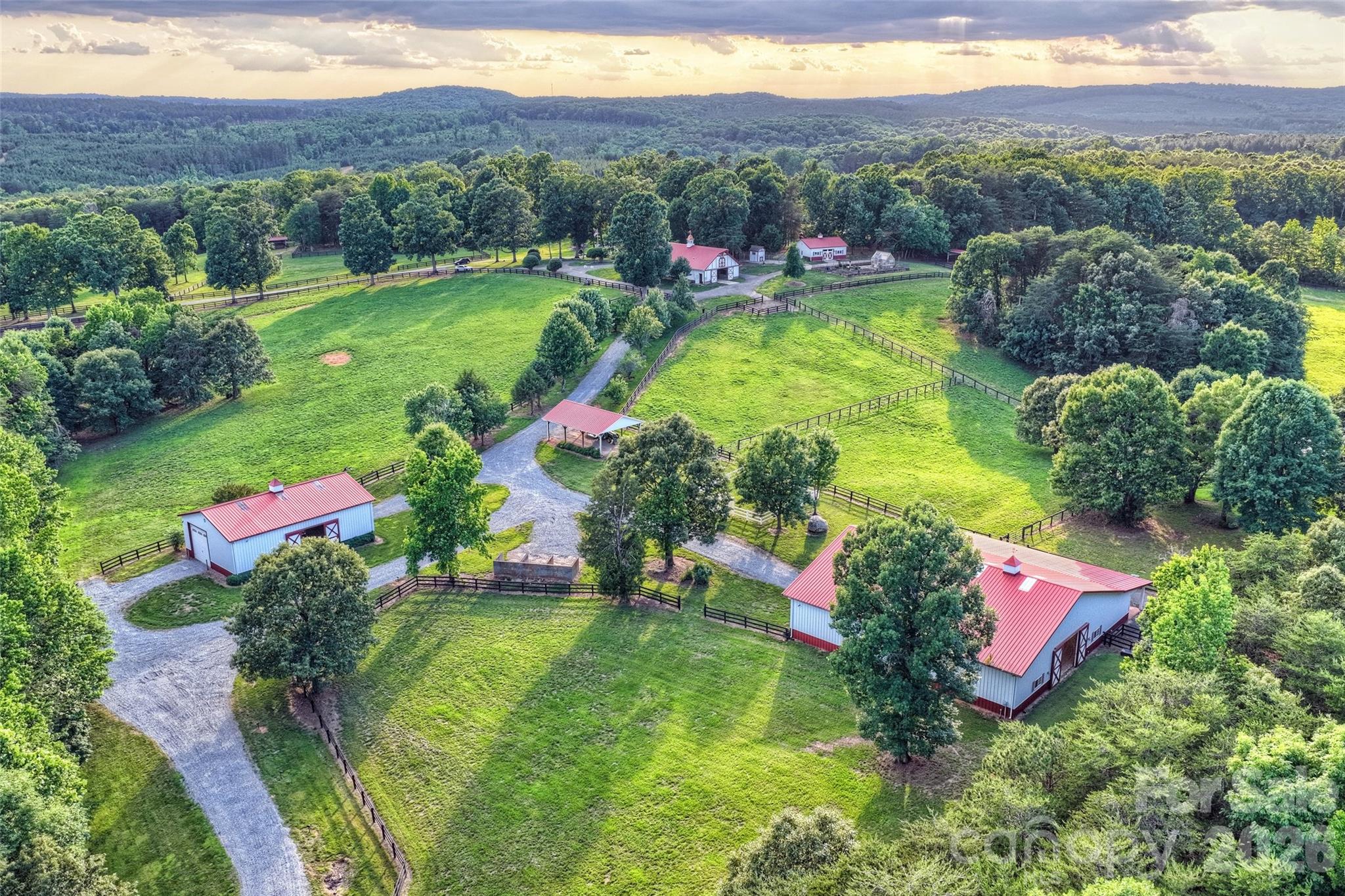 Overlooking rolling pastures and the Uwharrie National Forest, 172 Tuckertown Road commands attention with its Frank Lloyd Wright–inspired design, seamlessly blending architecture and nature through extensive stonework, rock waterfalls, natural landscaping, and exceptional indoor-outdoor living spaces.
A private gated entrance leads to the porte-cochère and expansive motor court, providing garage and covered storage while offering convenient access to the pool pavilion, gardens, and scenic walking trails throughout the property. The residence encompasses more than 7,000 square feet of thoughtfully designed living space featuring Spanish cedar columns and beams, soaring ceilings, a dramatic central atrium, sunroom, main-level guest suites, and walls of windows that capture the surrounding pastoral views. The lower level is designed for entertaining and extended living, offering additional guest suites, a home theater, fitness room, recreation areas, and direct access to the outdoor amenities. Resort-style outdoor living includes cascading stone waterfalls, koi ponds, a pool and spa, outdoor entertaining spaces, luxury dog kennels, and a  greenhouse surrounded by a charming English garden.
Equestrian and farm facilities are equally impressive. The main barn features seven stalls, a laundry area, full bath, wash facilities, tack room, and a fully furnished apartment above. The indoor riding arena offers sand footing, five stalls with Dutch doors, and year-round riding capability. Additional improvements include vehicle storage buildings, an aviary, chicken coop, run-in sheds, multiple accessory structures, and a service building with 14-foot doors ideal for RV, equipment, or workshop use. A rare offering that combines architectural significance, luxury living, equestrian excellence, and a private resort lifestyle in one extraordinary estate.