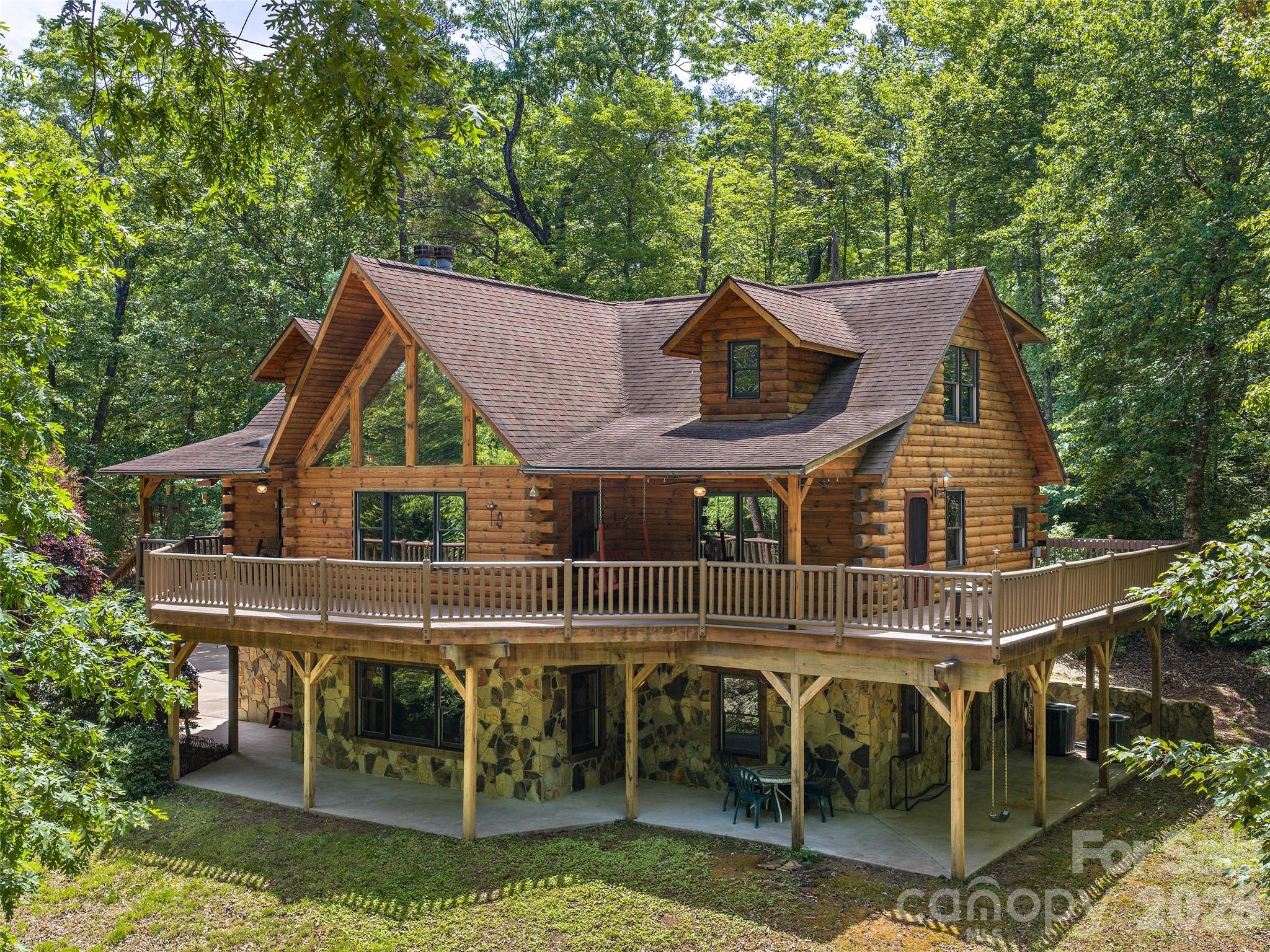Welcome to this stunning log cabin nestled in the sought-after Lake Adger community, offering the ultimate blend of rustic charm and modern luxury on 3.7 gently rolling acres. Crafted with authentic log construction and accented by beautiful Tennessee Crab Orchard stonework, this home boasts a timeless stone wall and exceptional curb appeal.
Enjoy outdoor living at its finest with an expansive wrap-around porch on the main level, a spacious ground-floor patio, and a private back deck—perfect for relaxing or entertaining while surrounded by the peace and tranquility of nature. Step inside to a grand great room featuring a soaring vaulted wood ceiling and a striking see-through stacked stone fireplace, creating a warm and inviting focal point. The chef’s kitchen is a showstopper, highlighted by a large granite island with a copper prep sink and breakfast bar, an antiqued 1800’s-style Elmira Stove Works 6-burner gas range with pot filler, additional wall oven, stainless steel appliances, ample cabinetry, a walk-in pantry, and charming antique accents. The spacious primary bedroom offers direct access to the outdoor deck, while the luxurious primary bath includes a walk-in shower and a relaxing Jacuzzi tub. The main level also features a versatile office or den, a guest bath, and easy deck access. Upstairs, discover a bright loft area, two comfortable guest bedrooms, and a full bath. The lower level is designed for fun and flexibility with a generous recreation room complete with a wet bar and kegerator, plus two additional bedrooms or flex spaces, another full bath, and direct walk-out access to the wrap-around patio. New Trane furnace and air conditioning were installed in 2025. Experience unmatched privacy, quiet, and natural beauty in this serene Lake Adger retreat. The adjoining 3.12 acre lot is also available for additional purchase to add additional privacy, to create a family compound, or to clear for horse pasture. Lake Adger Residents enjoy access to one of Western North Carolina’s most beautiful waterways, including a boat ramp, lake access, and a private community pavilion. Lake Adger encompasses 14 miles of shoreline and 438 surface acres of water, ideal for boating, kayaking, fishing, and swimming. The Lake Adger Community is a private, planned development covering approximately 3,250 acres, featuring 13 miles of private equestrian and hiking trails. This location is conveniently accessible from Asheville and Hendersonville, NC, as well as Greenville and Spartanburg, SC. Please make sure to check out the Video Virtual Tour for a closer look. Don’t miss your chance to own this extraordinary log home—schedule your private tour today!