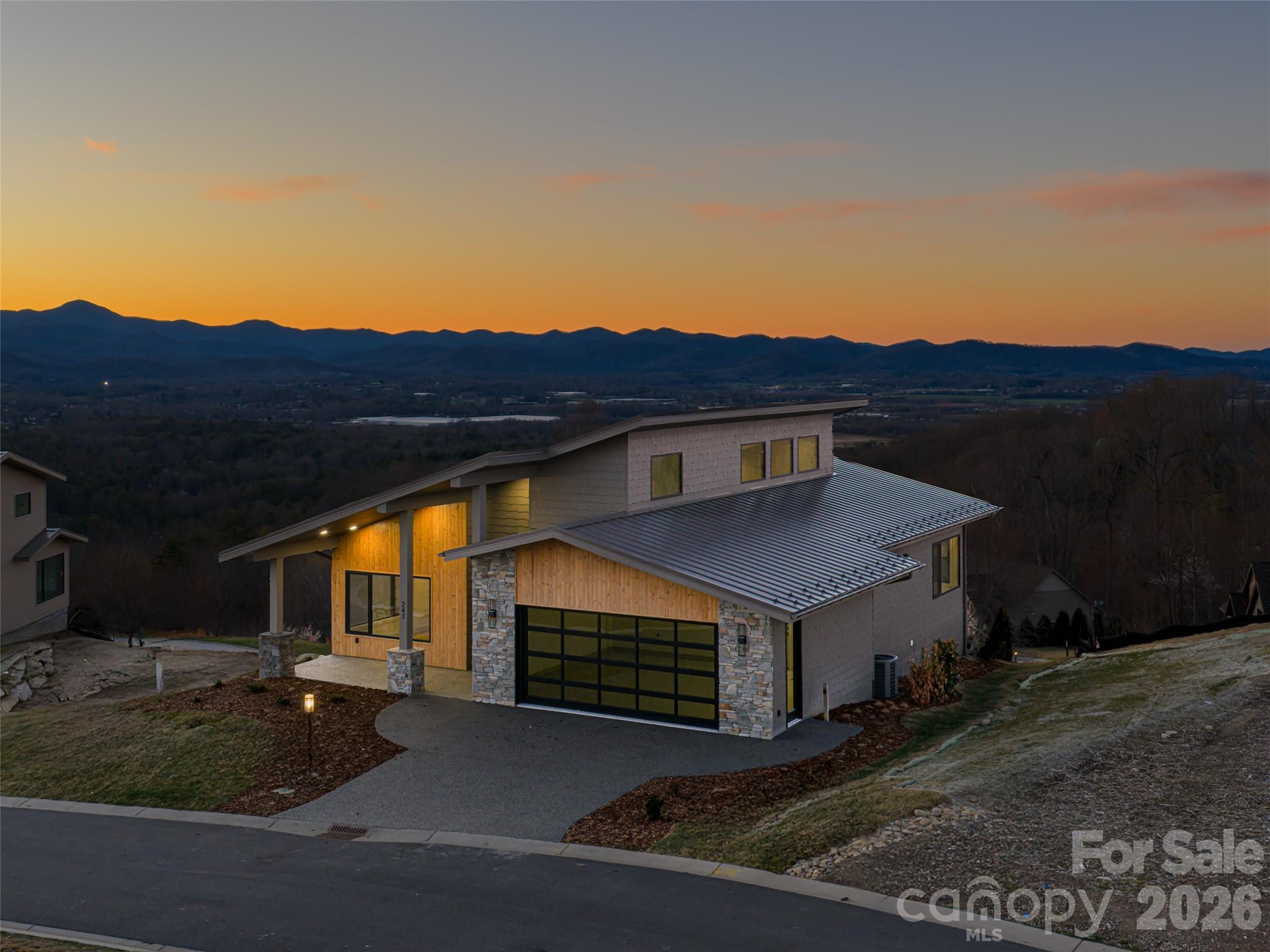 Perched within Summit Vista, the newest luxury enclave inside the gated Carriage Park community, this newly constructed mountain-modern home captures breathtaking, long-range mountain vistas from nearly every vantage point. Designed with a refined lock-and-leave lifestyle in mind, the residence offers exceptional privacy while remaining just 7 minutes from Historic Downtown Hendersonville and a short drive to Asheville Regional Airport. All Summit Vista homes are thoughtfully sited to live privately, yet remain naturally connected to neighbors and the greater Carriage Park community., including 6 acres of shared green space Inside, the open-concept main level is centered around a dramatic floor-to-ceiling stone fireplace, while expansive sliding doors open to a covered Garapa wood patio that showcases the sweeping mountain backdrop - an ideal setting for entertaining or quiet evenings outdoors. The chef’s kitchen pairs modern design with professional-grade performance, featuring elegant Calcutta Virtuoso quartz countertops and a full suite of Thermador appliances. The main-level primary suite offers a serene retreat with direct patio access and stunning mountain views, accompanied by a spa-inspired bath designed for relaxation. The spacious terrace level expands the home’s versatility with a second primary suite, additional guest suite, family room with wet bar, and a hobby or flex room with its own private entrance. A fenced yard on the terrace level, rare within the community, provides the perfect space for pets or private relaxation. Ready for your vision, the terrace level offers exceptional flexibility for guests, hobbies, or extended living. Residents of Carriage Park enjoy 24-hour gated security, a clubhouse, heated indoor pool, tennis and pickleball courts, and scenic common areas, making this an exceptional opportunity to own new-construction luxury in one of Hendersonville’s premier mountaintop communities.
