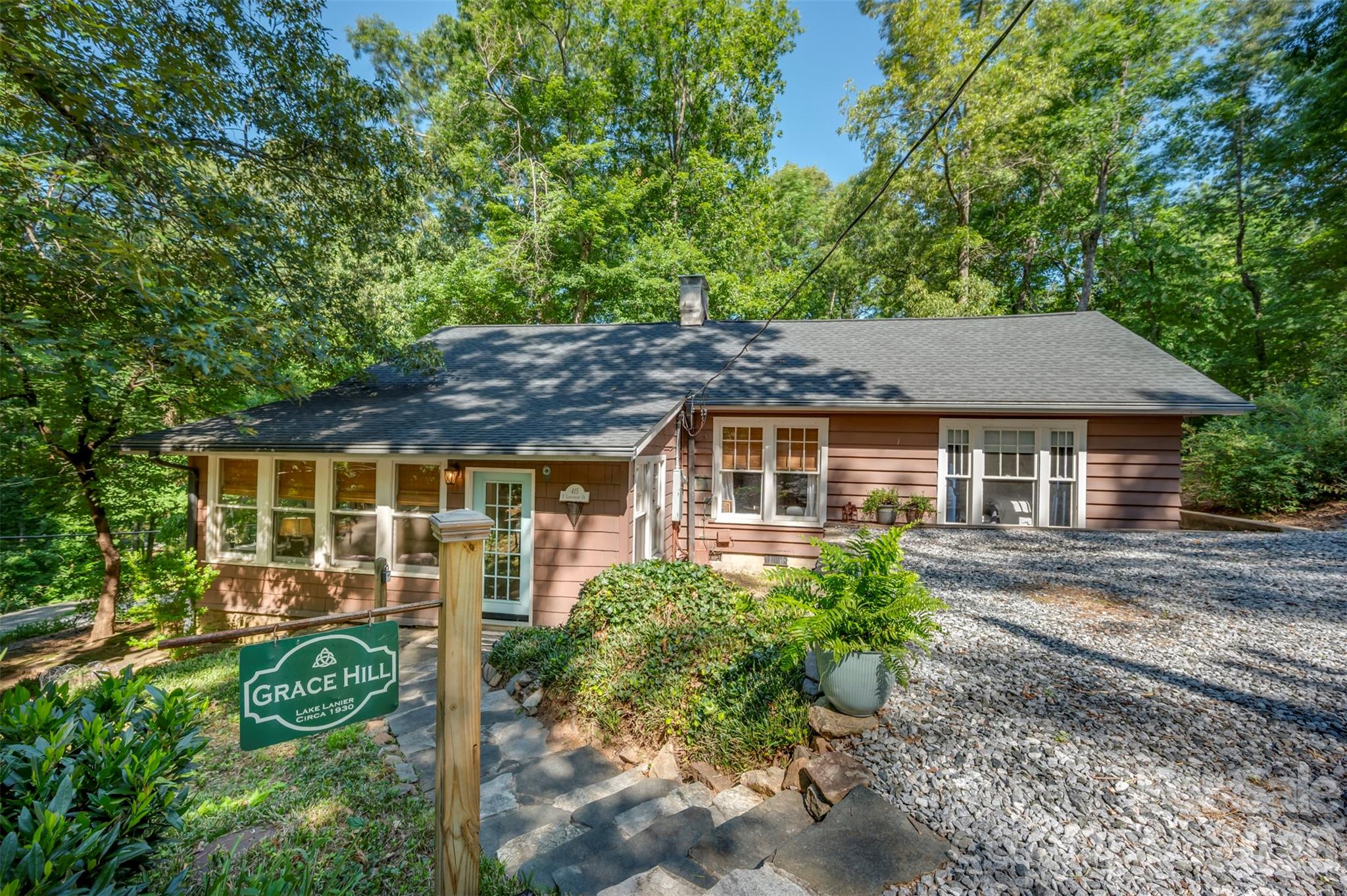 Grace Hill, circa 1930, captures the quiet charm of the Lake Lanier community — wooded streets, peaceful walks around the lake, water views along the way, and an idyllic rural feel just minutes from downtown Tryon and Landrum. Tucked beneath mature trees just off the water, this character-filled cottage offers the feeling of a private retreat with easy access to the everyday conveniences, restaurants, galleries, grocers, and entertainment venues of Tryon, Landrum, Columbus, and Saluda.
The cottage blends vintage character with relaxed livability: wood shingle and lap siding, a stone foundation, warm wood floors, a welcoming sunroom, bright cottage kitchen, and a comfortable living room anchored by a brick fireplace. The home’s informal layout, sloped-ceiling loft/bonus space, and cozy nooks give it the personality of a true lake-community cottage rather than a standard suburban floor plan.
Outdoor living is a major part of the appeal. Decks, a pergola, stonework, patio areas, and wooded surroundings create inviting places for morning coffee, evening gatherings, gardening, or simply enjoying the quiet. The setting feels tucked away, yet 415 E. Lakeshore is just about half a mile from the Lake Lanier dam, giving you a quick and easy route out to Hwy. 176. Nearby lake landing access is available, though there is no private dock or waterfront frontage. Buyers should verify lake access, parking, and practical use to their satisfaction. Grace Hill is a rare opportunity for those seeking history, character, privacy, and the relaxed rhythm of the Lake Lanier/Tryon/Landrum lifestyle.