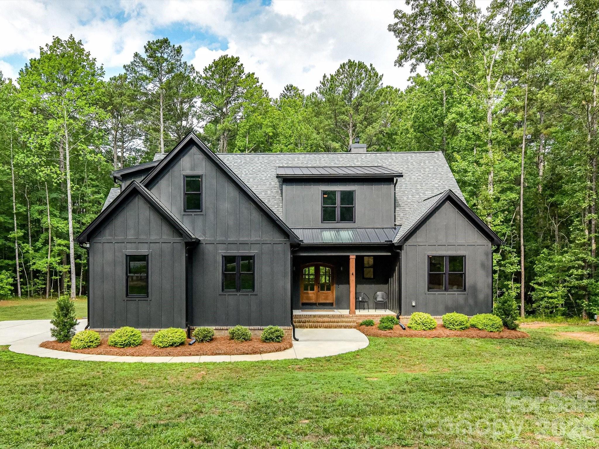 Privacy meets convenience in this custom 2023 modern farmhouse, set on a secluded 4-acre wooded lot in the Van Wyck community— yet less than 15 minutes to shopping, dining, medical facilities, and everyday essentials in Indian Land, plus the historic charm and downtown energy of Waxhaw. A cleared 1-acre homesite is bordered by 3 acres of mature woods, and the lot's natural slope frames elevated, treehouse-like views of the surrounding canopy from both the interior living spaces and the outdoor porches — a rare sense of seclusion just off Charlotte Hwy near the NC border.
The exterior is a study in craftsmanship: rich charcoal board-and-batten and lap siding rise above a handsome brick foundation, framed by cedar entry columns and a double-arched wood-and-glass front door beneath a standing-seam metal roof accent. Around back, a screened porch and open covered deck sit elevated among the trees, connected by a staircase that descends into the yard below — every detail, from the brick chimney to the black-trimmed windows, reflects the same level of care found throughout the home. 
Step outside the great room to a true extension of the home's living space: a screened porch and covered deck overlook the wooded homesite; the covered deck is anchored by a wood-burning brick fireplace that creates a year-round retreat for entertaining. Large windows and an 8-foot sliding glass door frame the wooded views from inside, blurring the line between indoors and out.
An expansive kitchen island anchors the open-concept main level, featuring solid-wood soft-close white cabinetry, an undermount porcelain farmhouse sink, and honed black granite counters, a favorite of the sellers! A gas range and modern stainless exhaust hood serve as the focal point, set against a striking black matte herringbone subway tile backsplash. The main living area rises from 9-foot ceilings to an 18-foot cathedral height detailed with a center wood beam, filling the space with light and treetop views. Truly a beautiful space to gather with friends.

The main-level split-bedroom plan includes 3 bedrooms, highlighted by a primary ensuite with a soaring 16-foot cathedral ceiling. The primary bath offers a walk-in shower, freestanding soaking tub, dual vanities accented with shiplap backdrop, a private water closet, and classic black hexagonal tile flooring. Upstairs, a spacious 16x23 bonus room and a private 4th-bedroom suite with a full tub-and-shower combination provide flexible space for guests or additional living needs. Storage is abundant, with an oversized 2-car garage, a mudroom drop zone, a walk-in attic, and multiple secondary attic spaces. In addition, there is a prewired Cat5 network hub, ideal for seamless connectivity. The home is heated and cooled by 2 HVAC units. 
NuCore luxury vinyl plank flooring runs throughout the main level, paired with black farmhouse lighting fixtures, custom blackout shades, and black ceiling fans. 
This home offers an ideal transitional setting — the tranquility of wooded, private acreage paired with quick access to the growing conveniences of Indian Land and the urban vibes of Historic Waxhaw. Schedule your private tour today to experience all of the custom details this property has to offer first-hand.