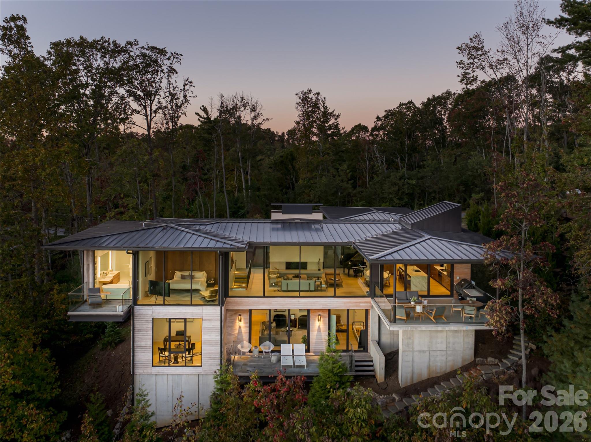 Perched along a dramatic ridge north of Asheville, this award-winning modern residence captures mountain serenity through architectural mastery. As clouds drift and the sun sets behind the Blue Ridge, floor-to-ceiling glass reveals sweeping views of layered peaks and the twinkling lights of downtown Asheville. Designed by Scott Huebner of Harding Huebner Architects, the home is a striking union of strength and transparency, conceived around a single sweeping roof and floor plane that suspends an all-glass core above the rugged terrain. Steel and concrete anchor the design while enhancing its sense of weightless elegance. Set on a half-acre lot, the 3,330-square-foot home achieves remarkable openness through intelligent orientation and engineering. A cantilevered terrace extends over the slope, offering panoramic views and unforgettable sunsets. Natural materials such as unstained wood siding, locally quarried stone and regionally sourced sand, create a seamless connection to the landscape. Inside, 11-foot ceilings, expansive windows, custom walnut cabinetry, polished marble surfaces, blonde oak floors and refined neutral tones balance warmth with modern minimalism. Honored with multiple design awards, including the AIA Asheville Honor Award and national Best in American Living recognition, this residence represents an elevated expression of contemporary mountain living, where architecture, light and landscape exist in harmony.