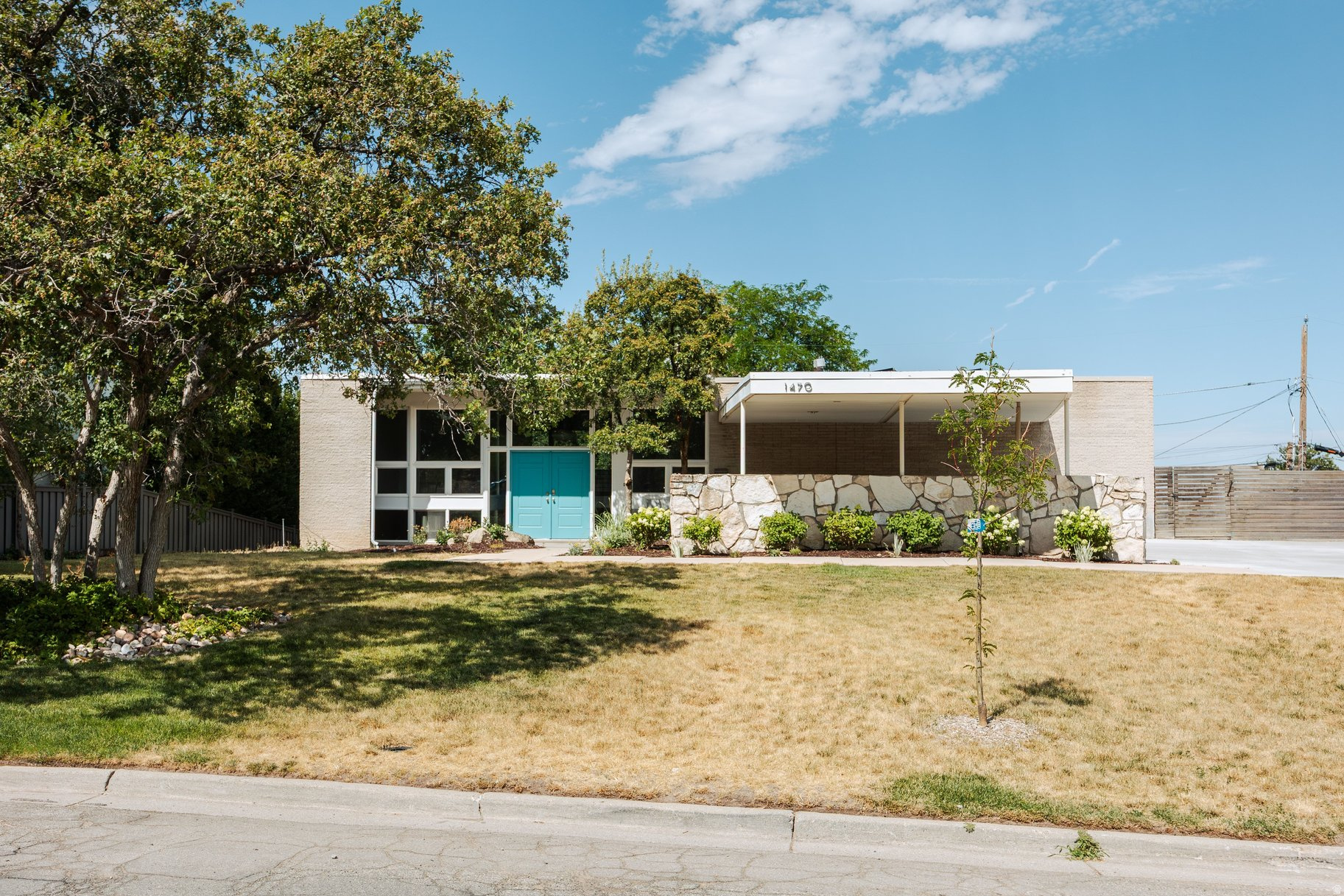 I absolutely love carports; there's something so wonderful about them that beautifully captures the midcentury allure of car culture. The image of classic cars with elegant wings sitting under the structure is not only highly pragmatic but also adds such a gorgeous detail to the home's architectural lines. This home features a wonderfully simple design, highlighted by a cantilever deck that hangs on the back side like a sculpture clinging effortlessly to the structure. Instead of running the entire length of the back, it intentionally creates a lovely void at one end. For such a clean, simple design, this gives the home that perfect little flair and a delightful design nod to a clever feat of engineering. What's truly fascinating about this home is how the living space is situated, which is something I rarely see! It offers a unique connection between the indoor and outdoor spaces. The main living areas, including the kitchen and dining room, are located on the lower ground level at the back of the home. This means that when you're in the kitchen, the beautiful backyard is just a sliding door away! It's such a wonderful idea to connect the public space this way, leaving the upper level perfectly private for the bedrooms and stunning city views.