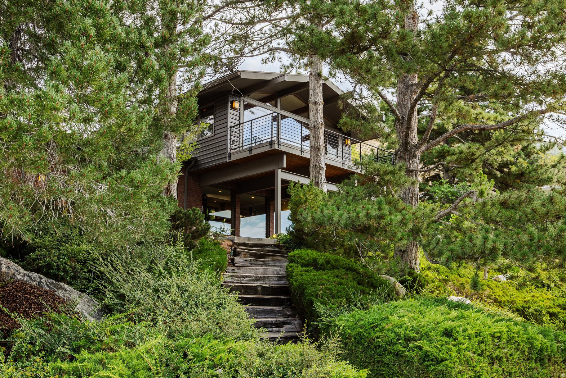 Some homes are designed to occupy a site. Others are designed to become part of it.From the street view, this home perches like a bird up high, shrouded beneath a cover of trees.Nested into the natural hillside at the top of the Upper Avenues, this woodsy mid-century contemporary embraces its surroundings with timeless architectural intent.Native stone, towering evergreens, and sweeping foothill vistas create the illusion of a secluded mountain retreat just minutes from downtown. Aweatheredwood staircase carves a path through the lush greenery,offering guests an unforgettable approach to the front entrance. For convenience's sake, a circular driveway leads to a three-car garage, plus additional sheltered parking -- a real rarity in the Avenues.Expanses of glass, soaring vaulted ceilings, and exposed structural beams create a natural dialogue between indoors and out, while generous decks extend the living spaces into the landscape. Natural light filters through the trees throughout the day, changing with the seasons and bringing nature into nearly every room. Thoughtfully positioned tomaximize both privacy and views, the home embodies the enduring principles of mid-century design-clean lines, honest materials, and a deep connection to its setting. Whether enjoying quiet mornings beneath the pines or gathering against the backdrop of the surrounding foothills, this is a home that encourages earthing and a greater reverence for the landscape.A private basketball court behind the home offers space for recreation today, or perhaps the perfect excuse to discover your new favorite pickleball court. This special home provides you the unmatched character of the historic Avenues, with backdoor access to the beautiful Bonneville Shoreline trail system. I'm bound to see you there.