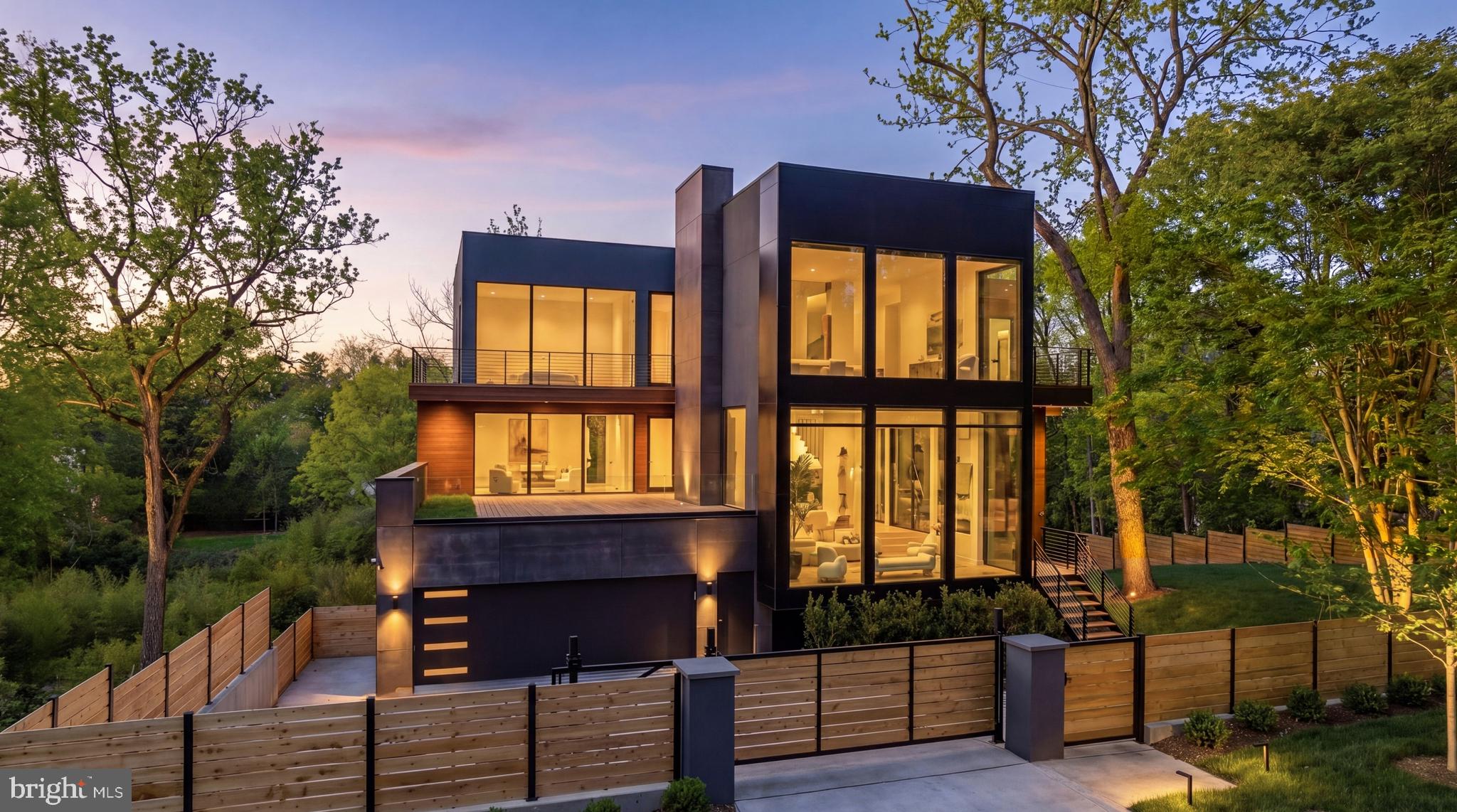 A bold architectural statement set against verdant parkland, 2676 Chain Bridge Road NW is a newly constructed modern estate designed by Patrick Brian Jones and built by Thorsen Construction. Positioned at the intersection of sustainability and harmonious site integration, the residence unites refined contemporary design with advanced building systems, resulting in a home of striking presence and enduring performance.

Set behind automatic gates on a coveted 0.46-acre parcel bridging the Berkley, Palisades, and Wesley Heights communities, the property occupies one of Washington’s most sought-after locations. Integrated into the natural grade, the exterior is defined by a considered interplay of thermally modified wood siding, viroc concrete panels, and insulated aluminum panels, complemented by expansive walls of thermally broken aluminum-framed glass by Western and Windsor Window Systems.

Beyond a pivoting mahogany solid-slab front door, soaring ceilings establish a sense of volume and light, allowing daylight to cascade across every level. Surrounded by glass, the great room is anchored by a linear fireplace feature wall and illuminated by a cascading raindrop chandelier. The dining room balances formality with everyday livability, framed by sliding doors that open to wooded surroundings. A glass-enclosed, temperature- and humidity-controlled wine room with custom glazing and bespoke racking serves as a sculptural focal point.

The chef’s kitchen features dual islands, custom walnut cabinetry, and a professional-grade appliance suite. A cantilevered breakfast nook sits within a bay window, while the adjacent family room opens seamlessly to the outdoors. A private office with a full bath on the main level offers flexibility as a multigenerational suite. A fully integrated Lutron lighting controlled and shade system is concealed within the ceiling plane, preserving clean sightlines, while integrated speakers and a private elevator reinforce the home’s seamless union of design and technology.

At the top of a glass-enclosed staircase, the primary suite appears to float among the treetops, with a private balcony and a spa-inspired bath featuring a custom walnut vanity with double sink, separate makeup counter, veined marble, a frosted glass shower, and a freestanding tub set before an oversized window. A double-height dressing room with a floating island and custom cabinetry evokes the refinement of a private atelier.

Two secondary bedrooms share a sitting room and offer ensuite baths, walk-in closets, and private balconies. A generous in-law suite includes a private kitchenette, while a second office completes the upper level.

The lower level introduces a guest suite with its own kitchen, alongside a wellness center with a massage room, spa bath with sauna and steam shower, and a fitness room opening to the pool deck. A three-season room extends indoor-outdoor living, while a mudroom connects to the subterranean three-car garage.

Outdoors, a resort-caliber lap pool and spa are complemented by an expansive terrace and deck with a large retractable awning, creating multiple settings for al fresco entertaining with an outdoor grilling station.

Engineered for efficiency, the home features geothermal hydronic radiant floor heating throughout, along with a fully integrated solar and battery infrastructure. The system includes a 40 kW solar array with over 100 panels, a 20 kW Enphase battery backup, and an installed Level 2 electric vehicle charger, all supported by a new 800-amp electrical service. Ownership of the system conveys full control, energy independence, and long-term cost efficiency.

Executed with precision and vision, 2676 Chain Bridge Road NW stands as a rare modern estate defined by equal parts sustainability and sophistication, offering a future-ready lifestyle in one of Washington’s most coveted settings.