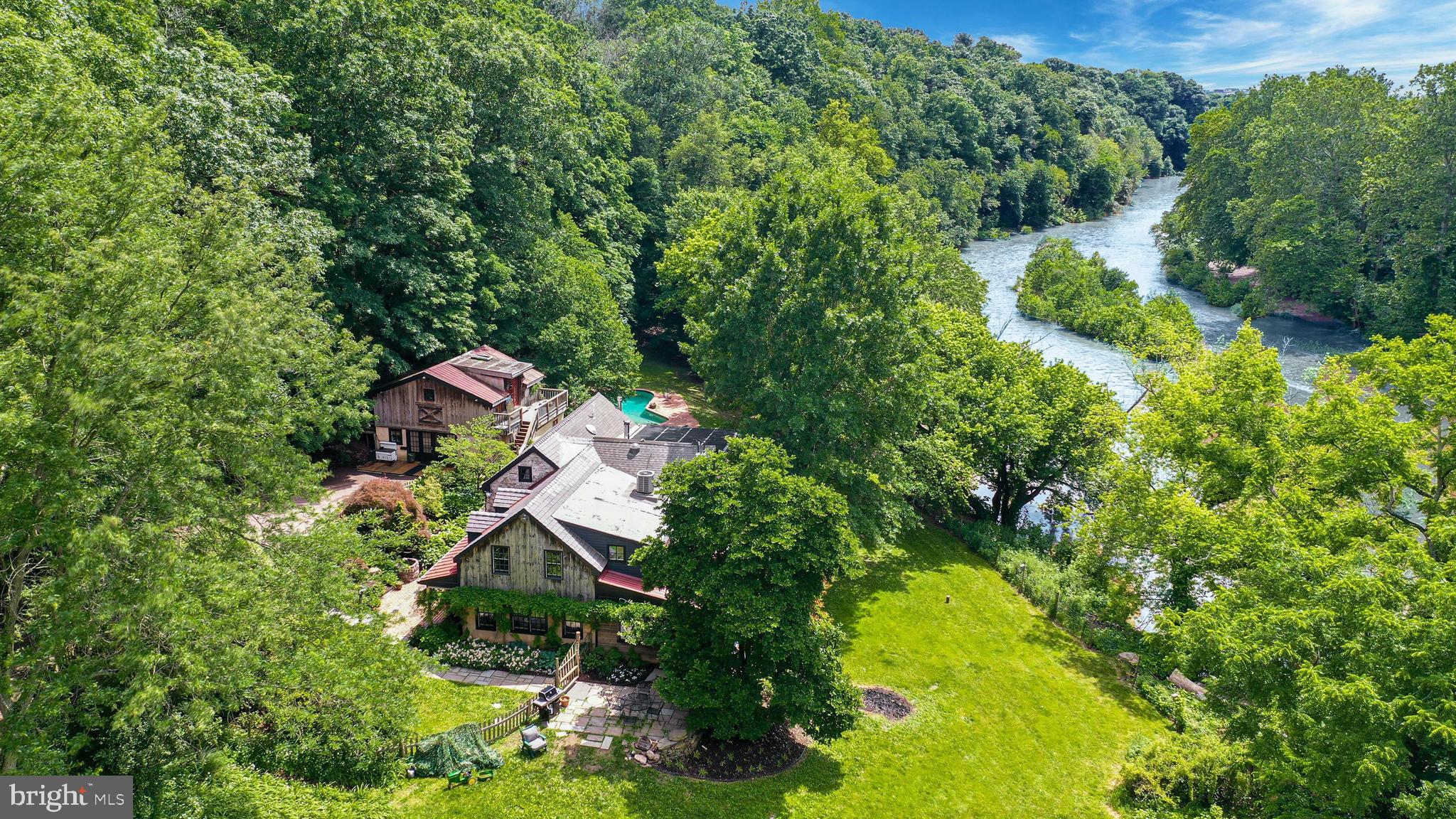 A winding gravel drive traverses the southeastern bank of the Perkiomen Creek, leading to this idyllic rural farmette dating back to 1708. The charming main house is surrounded by six acres of woodland, cleared meadows, a guest cottage, barn, and inground pool, all overlooking the ever-changing beauty of the creek as it flows by.

Rich in character and comfort, the house is welcoming with wood and tiled floors, deep windowsills, and custom updates throughout using reclaimed and repurposed materials. A cook’s kitchen is centered between the two sides of the home, with a walk-in stone fireplace that houses a 6-burner gas stove. Doors and windows open to the outside, and a sunroom and attached greenhouse take advantage of the natural surroundings in every season. Upstairs, four bedrooms and 3 full baths look out onto the lush landscape. With a brick patio out front, covered porch in the back, and a wraparound deck with hot tub, outdoor living is just as inviting as indoors.
The nearby cedar cottage houses beautifully appointed guest quarters with a kitchen and full bath on the main level, as well as a large office space above, perfect for guests or visiting family. Across the wide lawn, the cedar barn includes a workshop and storage space. Nearby is a heated inground pool, with privileged vistas of the creek and the verdant countryside beyond, and a view of the stars at night. Geothermal heating and solar panels allow the property to coexist harmoniously with its surroundings.