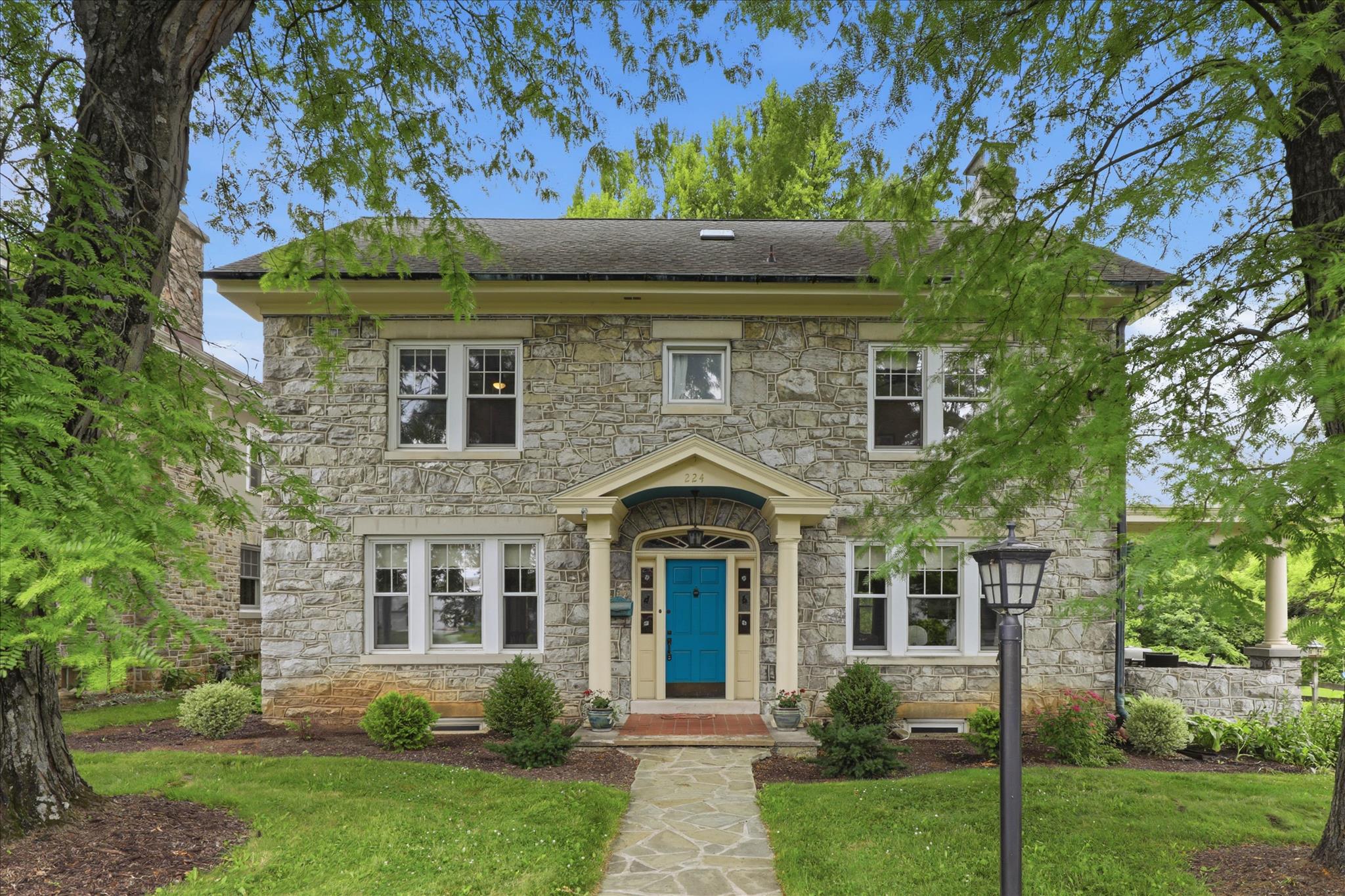 Some properties are built from the same material that shaped the land around them. 224 S 4th Street in Denver sits on .8 acres at the edge of where the borough gives way to Cocalico Creek, the water audible from the backyard, the pond with its fountain visible from the road. The fieldstone that clads the exterior is the same stone anchoring the floor-to-ceiling fireplace inside. This house was not assembled. It was quarried.
The entry sets the tone. A wide center hall with original hardwood floors, an arched front door surround with a sunburst fanlight, and millwork on the doorways that signals a standard of craft no longer practiced. Off the hall, a formal dining room with a crystal chandelier, cast iron radiators, and a built-in china cabinet with arched glass doors. The living room is anchored by the stone fireplace, flanked by French doors and large windows that frame the mature tree canopy outside. Six bedrooms and six baths spread across more than 3,000 square feet. The kitchen has a gas range, double wall ovens, and a farmhouse sink set beside built-in pantry shelving that runs floor to ceiling.
Back outside, the property runs deep. A large paved driveway. A covered stone porch. Then the lawn opens toward the pond and the creek, the kind of yard that takes a minute to walk end to end, framed by trees and Lancaster County farmland beyond.
224 S 4th Street represents a category of Lancaster County property (stone, scale, and setting) that is not rebuilt. It comes available as-is, or not at all.