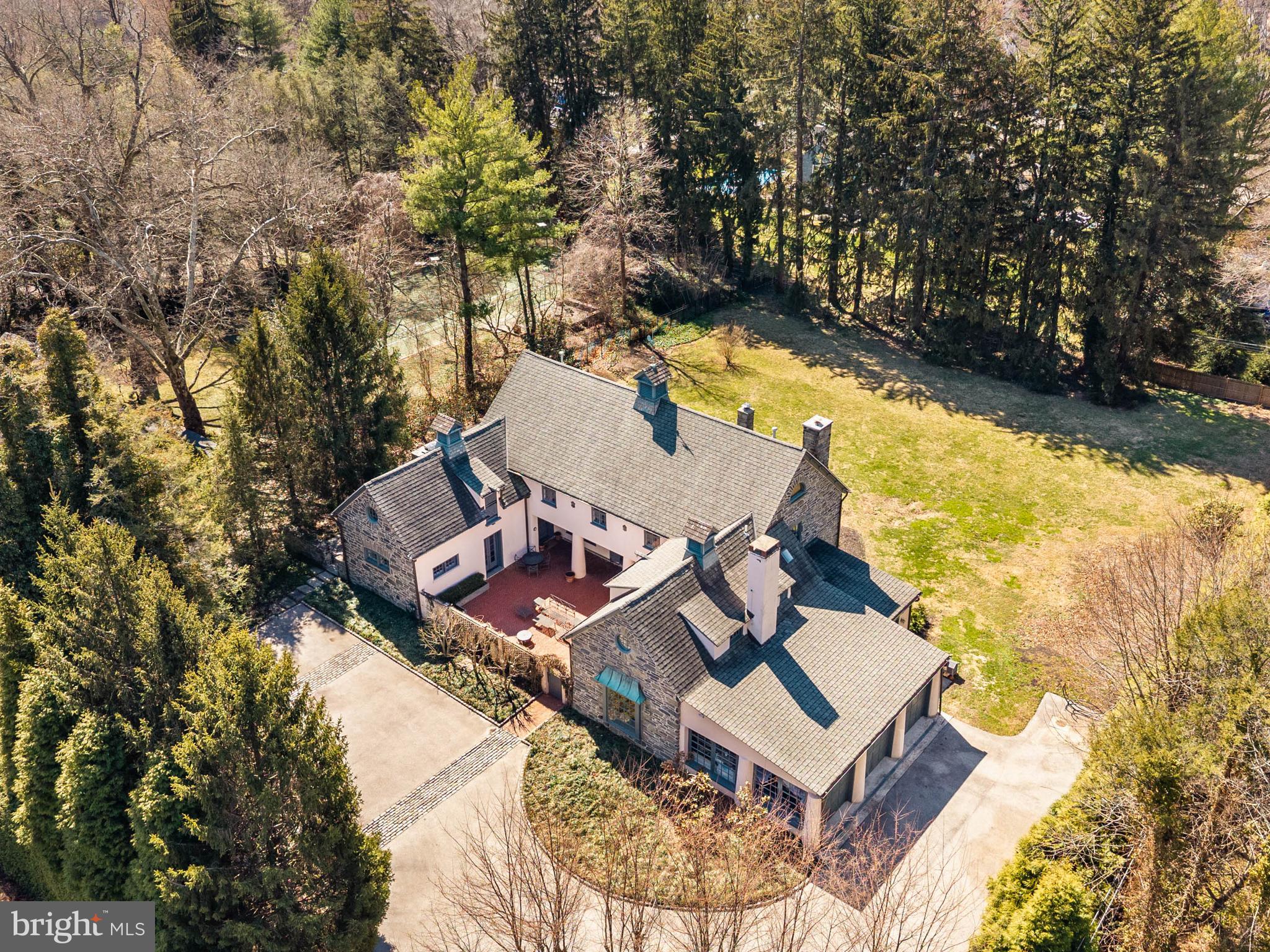 Stone pillars and a carved “Garden House” plaque mark the entrance to this exceptional 1.67-acre Main Line Carriage House, where a long tree-lined drive curves toward a house that rewards attention before you reach the door. The architecture tells its own story: the front elevation reads as a French manor — stucco and stone with a grand roof profile, three cupolas, and copper downspouts — while the rear presents a classic Main Line stone Georgian, symmetrical and grounded. A graceful, curved staircase with iron balusters and a wood runner anchors the stone-floored entry hall, setting a tone the rest of the house sustains through hardwood floors, true divided light windows with deep sills, original interior doors, and period woodwork throughout. The formal living room features full paneled walls, a wood-burning fireplace, floor to ceiling built-in bookshelves, wet bar, and French doors opening to the walled brick courtyard. The library sits adjacent with floor-to-ceiling built-in bookshelves, original woodwork, deep sill windows, a second wood-burning fireplace, and two sets of French doors to the courtyard and the rear patio — a room that becomes a passageway between outdoor spaces when the doors are open in summer. The dining room seats twelve comfortably with a large picture window framing the courtyard and is connected to the kitchen through a butler’s pantry and breakfast room. The kitchen is fitted with a Thermador range, SubZero refrigerator, Bosh dishwasher, and inset cabinetry and granite countertops, with a pantry, wine cooler, freezer, and laundry area providing direct access to the bluestone side patio and built-in grill. A separate family room off the kitchen opens to a second rear bluestone patio with morning sun and direct backyard access. The back entry includes a generously sized mud room — with an exposed stone wall and built-in bench and closets — and reads as a proper room before you reach the main entry hall. There is a half bath with original stone floor and seating area adjacent. Upstairs, the primary suite has a vaulted ceiling, porthole window, and built-in bookshelves, connecting two walk-in closets with custom built-ins and a primary bath with marble, soaking tub with jets, full marble tile walls, double vanity with marble countertop, and mosaic tile floor. Four additional well-proportioned bedrooms with original hardwood floors share two full baths, and a media room with built-in entertainment wall, sloped ceiling, and a second porthole window anchors the upper level. Adjacent, there is a laundry room with built in cabinets, bookcases, and linen storage. A full unfinished attic with high ceilings running the entire length of the house represents a significant expansion opportunity that most buyers will not anticipate. The property was originally part of the Colmar Estate, and the carriage house carries that history with a comprehensive renovation in 2005. The private grounds include a walled brick courtyard with decorative pillars for three-season dining, bluestone patios with a flagstone grill base and English bluestone herb garden, and a large flat rear lawn fully screened by mature Norwegian pines and over fifty Belgian arborvitaes. The house is oriented to catch morning sun in the front and evening light on the back patio, the gardens sequence through spring and summer in succession, and the property draws birds, foxes, and the kind of wildlife that signals a properly mature landscape. Three-car garage, Belgian block lined motor court, central air with new compressor (2025), public water and sewer, natural gas, in-wall and ceiling speakers throughout interior and courtyard, two laundry rooms, 5 bedrooms, 3.5 baths. Minutes to public and private schools, Merion Cricket Club, Suburban Square, Ardmore Station, and routes to Philadelphia and New York — a private setting with proximity to everything.