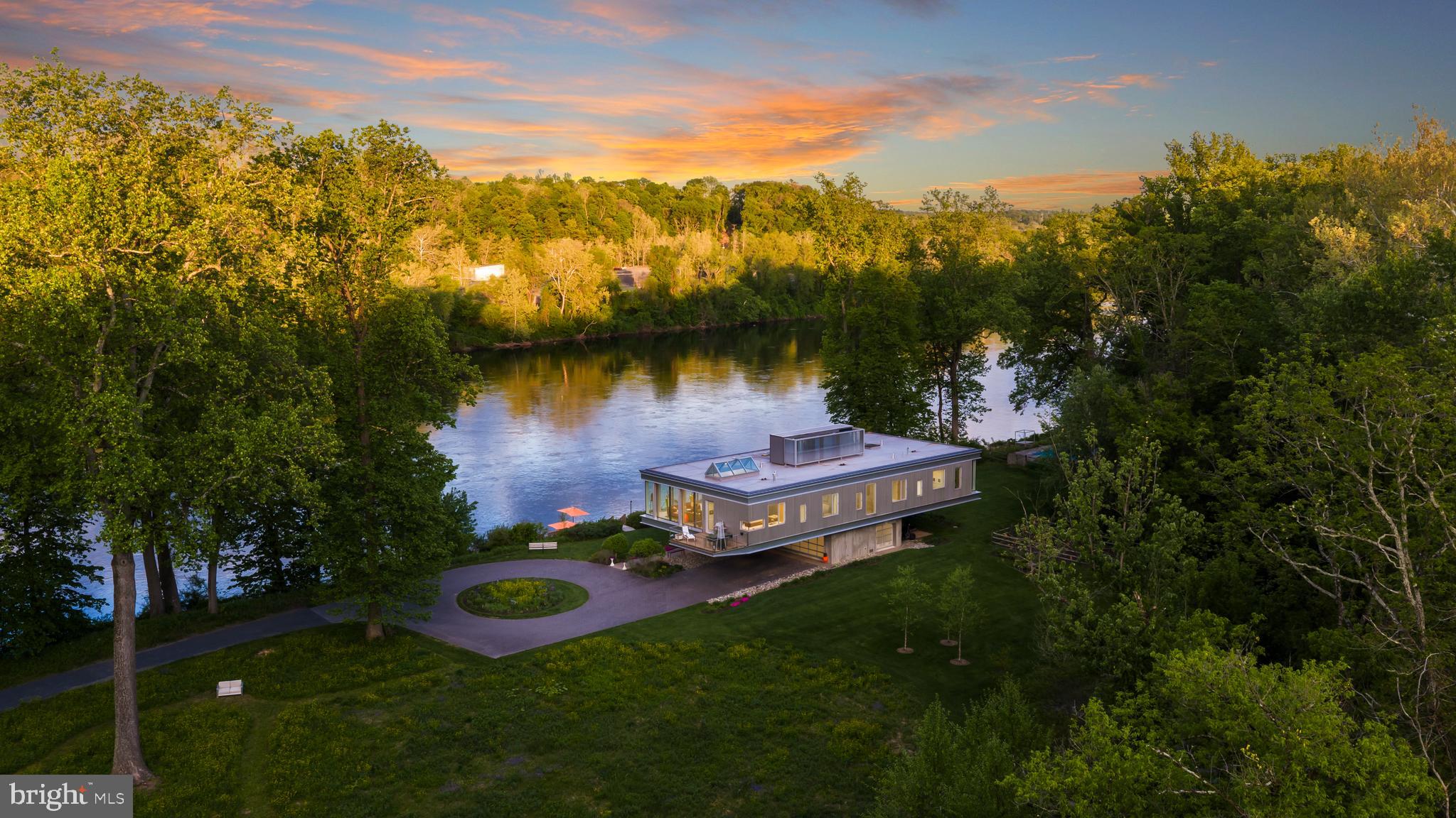 Riverscape is a rare architectural estate lifted gracefully above the Delaware River on over eight private acres, offering more than 1,100 feet of frontage in coveted New Hope. Perched like a sleek, floating pavilion above the floodplain, the residence is supported by deep helical piles, a 141‑piece structural steel frame, and three 14‑foot‑high trusses that cantilever the home over a sculptural poured‑concrete pedestal on the original 1930s cottage footprint. This base houses a three‑car garage and dramatic double‑height foyer with winding staircase, while engineered vents allow water to pass safely through in the event of flooding—an elegant, highly regulated solution for riverfront living.
The main living level unfolds in glass and light, framed by an Arcadia aluminum curtain‑wall system and ModernVu low‑E, argon‑filled casement windows that capture cinematic river and woodland views from full‑height walls of glass and two entertainment decks. An open kitchen and great room, finished with Mozambique cabinetry and Italian porcelain tile, form the heart of the home. The seductive primary suite—with two spa‑style baths featuring Italian marble, zero‑entry showers with linear drains, Grohe and Hansgrohe fixtures, and Toto toilets—acts as a private observatory for the ever‑changing water and sky. Three additional tranquil bedrooms, an exercise studio, and a teen lounge enjoy leafy, wooded vistas, creating a serene counterpoint to the river drama.
Every detail reflects a refined contemporary palette: exposed structural steel, board‑form concrete walls, polished concrete entry, polished stainless‑steel railings and cable, Walnut millwork, glass tile in secondary baths, Ipe decking, and aggregate walkways. Systems are equally elevated: radiant heat with supplemental linear‑diffuser hot air, wall‑hung boiler with stainless heat exchanger, two 200‑amp panels with whole‑house surge protection, Generac generator in its own shed above the flood zone, whole‑house Sonos with Sonance speakers, extensive coax and wiring, and a comprehensive Lutron platform with lighting control, wireless thermostats, smart‑tint windows, and motorized blinds. A TPO roof with metal flashing, VM Zinc wall panels on the penthouse, perforated stainless‑steel panels with high‑performance insulation, Hardie soffits, and closed‑cell insulation complete the highly engineered envelope.
Outside, the estate lives like a private resort. A gorgeous in‑ground pool with new pump, cordless vacuums, solar vacuum setup, and dedicated 50‑amp circuit for the hot tub anchors a series of stone‑terraced garden rooms designed for lounging and entertaining, all served by an irrigation system. A permeable paver circular drive, river rock installations, and a custom concrete entry sign introduce the property with understated drama. At the shoreline, a new cantilevered river deck with rotating benches, Roll‑N‑Go boat ramp, and private pathway to the towpath place sunrise paddles, afternoon bike rides, and moonlit cocktails just moments from your door. Wrapped in nature yet within easy reach of New Hope, Princeton, Philadelphia, and Manhattan, Riverscape is an impeccably designed, deeply sensual riverfront retreat and a once‑in‑a‑generation opportunity.