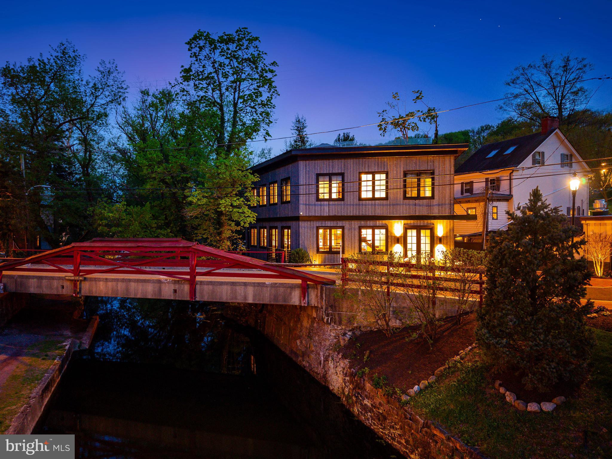 A rare opportunity invites you to own an architecturally brilliant home in the heart of New Hope, Pennsylvania. Perfectly situated on a charming canal street, this residence is naturally set apart from the lively Main Street by a tranquil canal. The signature red wooden bridge, one of New Hope’s most photographed landmarks, serves as your picturesque gateway to the town’s acclaimed restaurants, boutiques, and galleries. This newly constructed modern masterpiece showcases an inspired blend of European influence and contemporary design, drawing from Amsterdam and early modernist architecture. The interplay of light, space, and structure throughout its three levels creates a truly captivating living experience. Upon entering through the front entrance, a warm and inviting foyer welcomes you, with a striking staircase leading to the main living area. The open-concept design showcases gleaming hardwood floors and a refined sense of flow between each space. At the heart of the home is a beautifully designed wet bar with generous counter seating, perfect for entertaining. The living and dining areas exude modern sophistication, enhanced by expansive glass windows that fill the space with natural light. The sleek kitchen features black-grained European cabinetry, quartz countertops, a concealed pantry, and a substantial center island that anchors the room. Completing this level is an elegant billiard room with a custom built-in feature and a conveniently located powder room. On the second level, the full-floor owner’s suite offers the ultimate retreat with a spa-style bathroom, private terrace with a custom wet bar, office/study, laundry area, and a custom-upgraded closet system. Elegant window treatments, wallpaper accents, and designer lighting fixtures elevate each space with tailored sophistication. The lower level is equally impressive, featuring two additional bedroom suites each with its own full bath, a second laundry room, and a lounge area. Installed closets and upgraded storage systems enhance functionality and style. Every inch of the property has been thoughtfully curated, from the paver driveway and professionally landscaped grounds with lighting, to the artificial turf backyard overlooking the canal. Premium upgrades include a whole-house Generac generator, security and video system, triple sump pump system, whole-house carbon water filtration, an EV charger and a custom gutter system. Additional highlights include an elevator, three + private parking spaces, and direct canal access for a serene outdoor experience. With excellent proximity to both New York City and Philadelphia, you can enjoy New Hope’s vibrant lifestyle while living in your own private sanctuary. New Hope-Solebury School District is rated 10/10, and this address enjoys a premier in-town location surrounded by acclaimed dining such as Zoubi’s, Nektar, and Karla’s. Experience the best of luxury, design, and convenience, a home where every day feels like a holiday.
