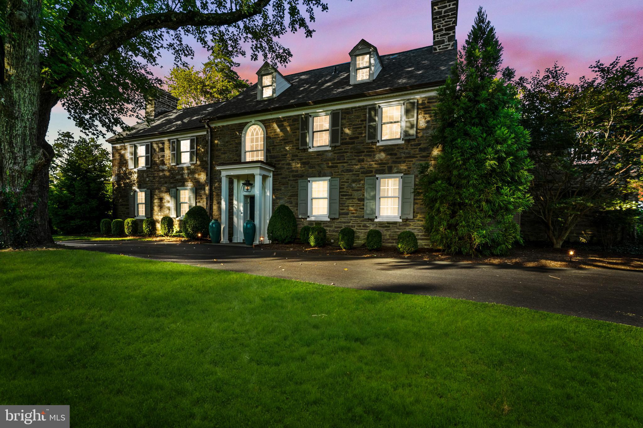 Classic and timeless, this Northside Haverford stone residence is gracefully sited on close to one acre of manicured grounds, where gardens, a shimmering pool, and tree-lined perimeters create a park-like setting. Approached by a circular drive that underscores its stately presence, the home reveals itself gradually balanced proportions, refined details, and interiors that have been thoughtfully modernized while honoring the architecture’s original spirit.

A step-up center foyer introduces a striking axial view: through the dining room’s sweeping bay window with its intricate glass roof, the eye is drawn directly to the gardens and pool beyond. To the left, the formal living room offers an atmosphere of understated elegance. An oversized fireplace, flanked by tall south-facing windows, anchors the space, while richly stained hardwood floors, continuous throughout the main level, add warmth and cohesion. Another bay window provides symmetry when viewed from the rear, and French doors open to a covered stone terrace—an invitation to flow seamlessly between interior and exterior living.

The kitchen is a study in contrast and comfort. Crisp white cabinetry plays against polished black stone countertops, while a substantial wood-topped island provides both prep and gathering space. A suite of Bosch appliances—including a steam/convection oven and dual dishwashers—makes the kitchen as functional for intimate dinners as it is for entertaining on a larger scale. Sunlight streams across the under-window sink, a small but significant detail that reinforces the room’s everyday ease.

For moments of repose, the den offers a more intimate counterpoint: custom shelving, front-facing windows, and a brick fireplace create a retreat equally suited to reading or quiet evenings by the fire.

Ascending the main staircase, an arched window becomes a focal point of architectural grace, as notable from the interior as it is when viewed from the façade. The primary suite, redesigned under current ownership, reflects both scale and serenity. The primary bath is anchored by a soaking tub positioned beneath a window, a spacious seated shower, and plenty of natural light from windows and large skylight.

A newly created walk-in closet, complete with its own heating system, offers ample storage with a sense of luxury and care.
Five additional bedrooms occupy the upper level—some connected by Jack-and-Jill baths, others with en-suite or adjoining arrangements—allowing flexibility and privacy. Anticipating the future, the attic has been spray-foam insulated, fully heated, and air-conditioned, ready for expansion should additional living space be desired.

On the lower level, daylight streams into a casual gathering area with fireplace and deep window wells—an inviting setting for informal entertaining. A secondary staircase connects this space to the kitchen, leading also to a large laundry area, half bath, and generous storage.

Outdoors, the home’s placement on its deep lot is deliberate and graceful. The covered terrace and poolside stonework blur the line between garden and architecture, while a recently re-roofed garden shed adds utility. A multi-car courtyard tucked to the rear offers discreet yet ample parking, underscoring the estate’s ability to welcome both intimate and grand occasions with equal ease.

Classic and enduring, the home is one of balance and beauty—bright yet grounded, sophisticated yet welcoming—an estate of timeless appeal in one of Haverford’s most distinguished enclaves