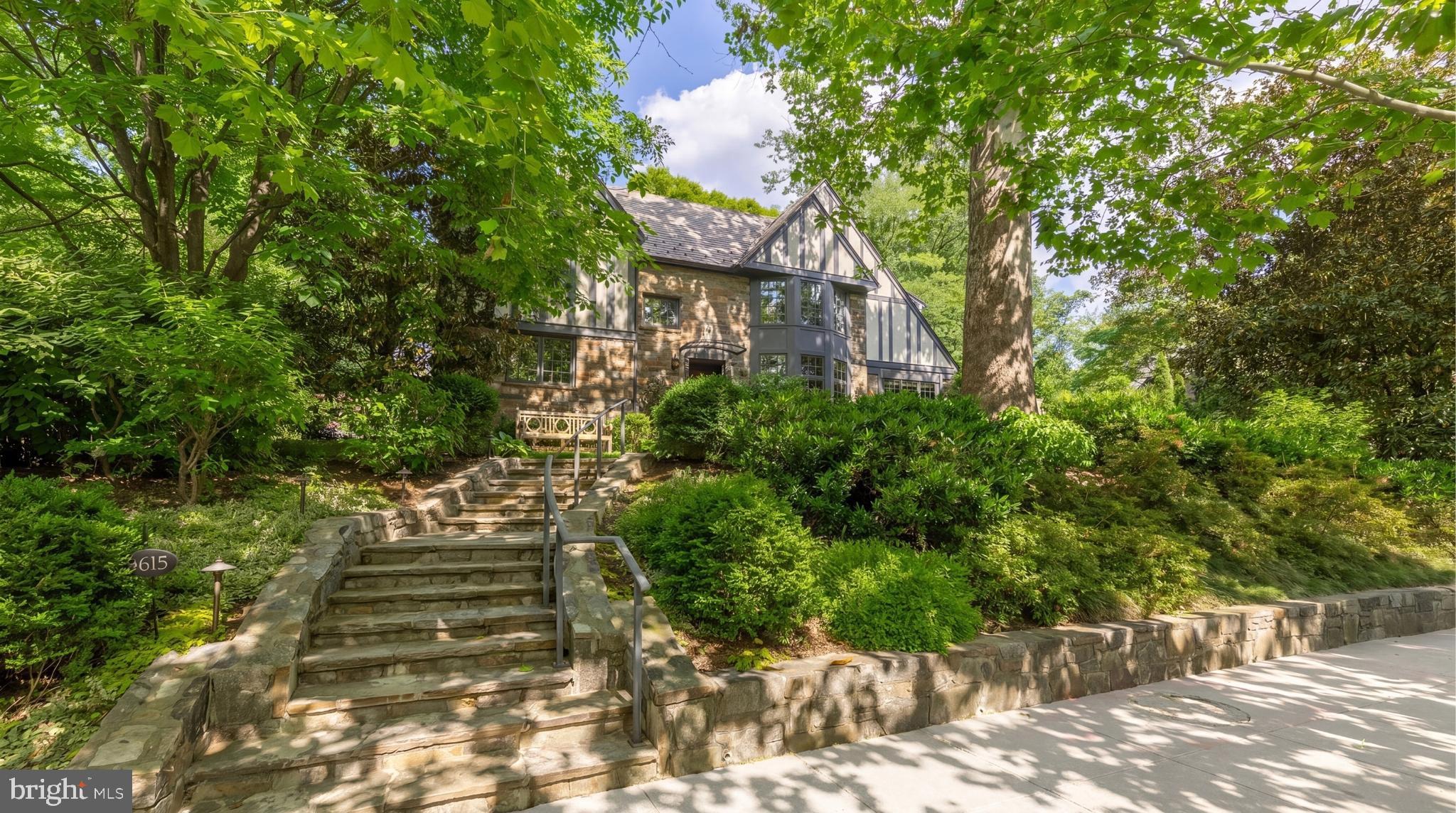 Set along one of Spring Valley’s most established streets, 3615 49th Street is a 1935 English Tudor extensively reimagined in 2012 by McLean-based Morris-Day Designers and Builders under architect Rob Morris. Spanning approximately 5,700 square feet, the residence is defined by architectural integrity, craftsmanship, and enduring character. Steep gables, half-timbering, textured masonry, and leaded windows preserve the essence of traditional Tudor architecture while introducing a more refined modern livability.

A stone stair ascends through layered gardens to a facade framed by climbing greenery and architectural detailing that feels timeless and established. Set within nearly half an acre, the surrounding landscape gives the home a notable sense of privacy within the city. Inside, the foyer opens to a series of formal rooms shaped by rich millwork, stained glass, exposed beams, and balanced proportions. The living room and library share a double-sided wood-burning fireplace, creating two connected gathering spaces, while French doors from the library open directly to the rear gardens and stone patio.

Throughout the main level, paneled walls, bespoke woodwork, and textured materials create warmth and architectural depth. A recurring rose motif within the custom interior glasswork lends a subtle signature to the home. The dining room is grounded by beamed ceilings and garden-facing windows, while at the rear of the residence, the family room rises beneath a cathedral ceiling with skylights and a gas fireplace, drawing inspiration from the great halls and ateliers that informed the renovation.

Designed for both everyday living and larger gatherings, the kitchen sequence centers around a classic AGA range, substantial cabinetry, a generous island, and a sunlit breakfast area. An adjoining scullery and secondary kitchen provide additional preparation and service space. From the rear living spaces, the home transitions naturally to an expansive stone terrace overlooking the landscaped grounds.

Occupying a private position on the second floor, the primary suite includes a spacious bedroom, walk-in closet, and marble bath with a soaking tub, dual vanity, walk-in shower, and beamed ceiling detailing. Two additional bedrooms share a Jack-and-Jill bath, with laundry conveniently positioned on the same level. The top floor introduces two additional bedrooms, a full bath, attic storage, and a walk-in cedar closet, creating flexibility for guests, office space, or creative use.

The lower level takes on the atmosphere of an old-world tavern, complete with a wet bar, fireplace, built-ins, ribbed ceiling detailing, and integrated seating. An exercise room, full bath, storage areas, and an extended mudroom connect directly to the attached two-car garage.

Moments from Spring Valley Park, neighborhood shops, restaurants, and top-rated schools, 3615 49th Street combines timeless design and day-to-day livability within one of Washington’s most established residential enclaves.