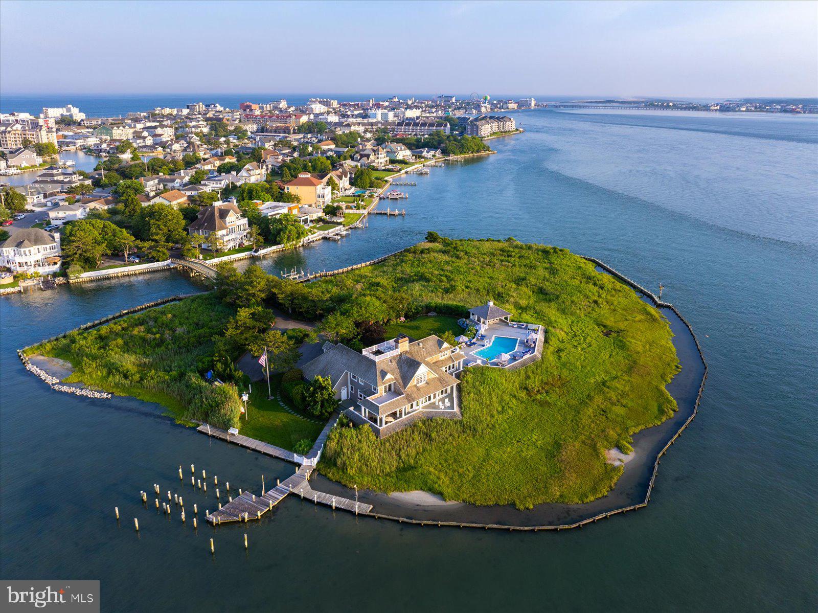 Goose Island at 1546 Teal Drive, Ocean City, Maryland is a truly unique offering — the bay’s only island estate. Located off 15th Street in downtown Ocean City, the property offers a remarkable balance of seclusion and accessibility,  3 blocks to the Boardwalk and beach, minutes to the Ocean City Inlet and Assateague Island by boat, and a block from Harbour Island, home of the world-renowned White Marlin Open. Hidden behind a brick wall extending along the property line and accessed through a 6-foot gate, the arrival experience continues across a private 120-foot vehicular bridge from Mallard Island. The island encompasses more than 3 acres of native marshlands and landscape with panoramic views of the open bay, the Rt. 50 Bridge to the south, the Rt. 90 Bridge to the north, and the rare combination of both breathtaking sunrises and sunsets across the water. Architecturally, the home is an iconic shingle-style residence. Inspired by Nantucket architecture, the residence underwent extensive exterior renovations and architectural enhancements under the direction of renowned Lyman Perry Architects Ltd. of Nantucket, Massachusetts, with Lyman Perry personally consulting on the estate. The approximately 5,000-square-foot home blends timeless design with exceptional craftsmanship, featuring custom woodworking, Carlisle wide-plank hand-finished pine flooring, new Andersen doors throughout, approximately 3,250 square feet of wraparound deck, and a resort atmosphere. Upon entering, a dramatic 24-foot atrium features an expansive 12-foot by 15-foot overhead skylight and a floor-to-ceiling 24-foot Coral Rock fireplace feature that anchors and connects the surrounding living spaces while filling the home with natural light. Designed to maximize open bay views throughout the home, the main living area features custom built-in cabinetry and entertainment center while flowing naturally into the separate dining and gathering spaces. The open-concept kitchen/family room floor plan creates an inviting living area designed for entertaining. Custom solid birch painted cabinetry by Premier Cabinetry of New Holland, Pennsylvania, includes a generous center island with a 5-foot by 8-foot teak countertop. A custom wood spiral staircase in the kitchen/family room provides secondary access to the upper level. The main level also includes a home office with built-in cabinetry and concealed pull-out file storage, along with an additional guest bedroom and full bath. Upstairs, an open bridge hallway overlooks the atrium and Coral Rock feature while separating the primary suite from two guest bedroom suites. The four-room primary suite consists of a bedroom retreat with access to a bayfront deck, a combined sitting, entertainment, and dressing room with vaulted ceilings, custom cherry wood built-ins and four closets, an en-suite bath with double vanity and dressing table, and a second dressing room featuring vaulted ceilings, four double-door deep closets, and an arched window with window seat dividing two built-in tall chests totaling 30 drawers. Two additional upstairs bedrooms each feature en-suite baths and private decks. Outdoors, the property transforms into an island retreat with a pool and spa by Sea Glass Pools, expansive decking, a large entertaining gazebo bar with cabinetry and quartz countertops, a full bath with shower, and protected docking facilities with parallel docking along approximately 1,600 feet of bulkhead, a 10,000-pound 2024 boat lift, and a fish cleaning station. Significant recent investments include bulkhead improvements, reinforced helical pilings, extensive bridge renovations, active security and surveillance systems, and meticulously maintained structural and mechanical systems. Surrounded by abundant birdlife including osprey, great blue herons, great white egrets, snowy egrets, mallards, and bald eagles, along with endless open-water vistas, Goose Island is a distinguished estate defined by timeless elegance.