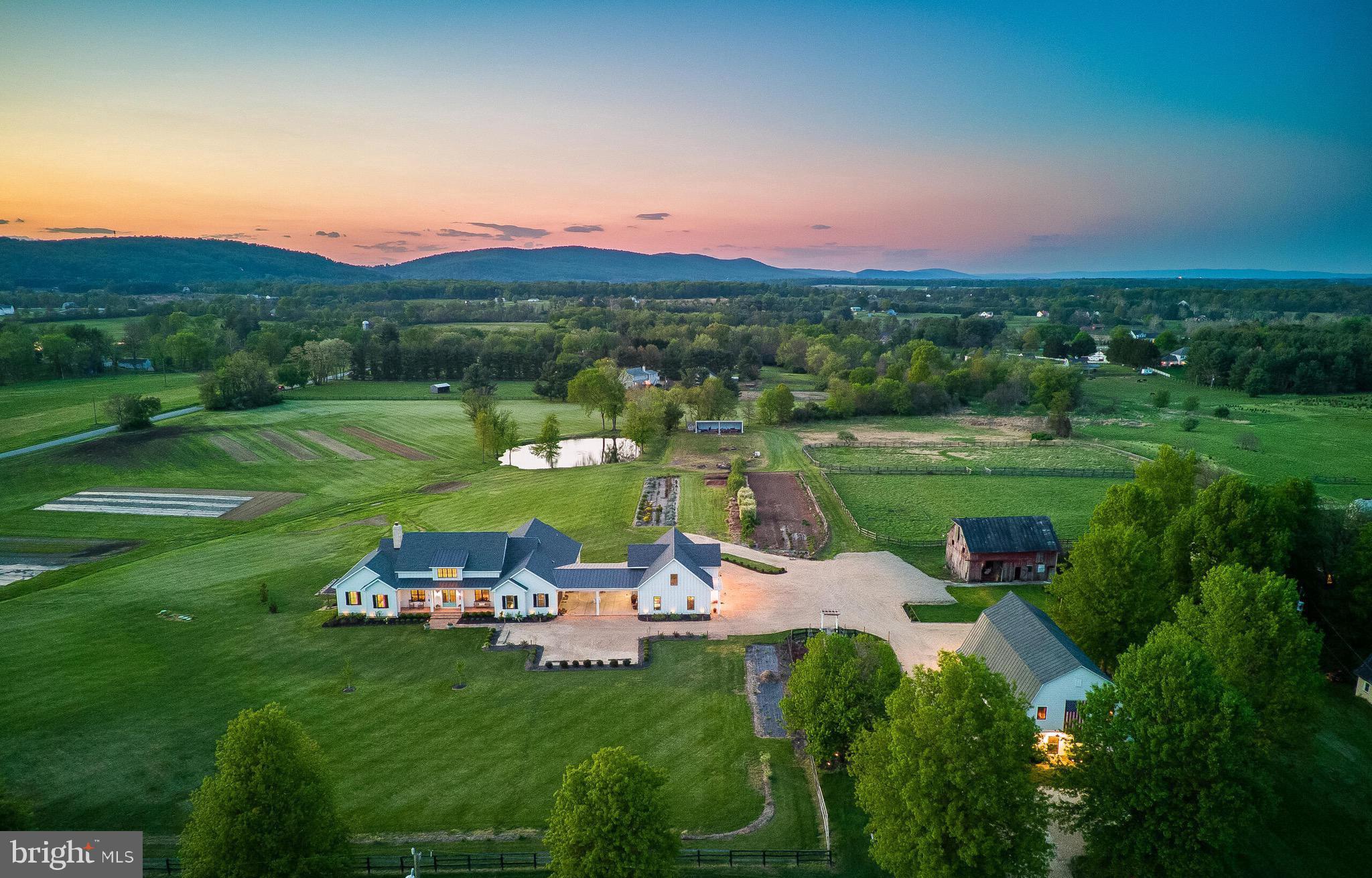 Blue Sky Farm was completed in 2023 as the personal residence of an established custom builder, and every specification reflects that singular standard. The residence is sited to harness the full drama of its natural setting — every principal room frames a living tableau of the pond, the long light of evening, and the silhouette of the Blue Ridge Mountains beyond. All four bedrooms occupy the main floor, arranged for both quiet separation and effortless daily flow. The lower level adds approx. 1,034 finished sq. ft. with a full bathroom, plus approx. 2,420 sq. ft. unfinished and ready for a future media room, gym, guest suite, or additional bedrooms. Total finished area is approx. 4,701 sq. ft.; total under roof, including outbuildings, is approx. 14,875 sq. ft. The grounds extend across approx. 15.33 acres on two parcels, encompassing a pond, commercial-scale perennial gardens, and the restored Blue Sky Barn with bathroom, kitchen, finished lower level and upper entertainment level with stage and bar.
Five minutes to Purcellville's restaurants, boutiques, and farmers market. Western Loudoun County is one of Virginia's premier agritourism destinations — surrounded by award-winning wineries, working farms, equestrian estates, and the cultural draw of Middleburg and Leesburg. A coveted address that feels worlds away from everything, yet isn't.
CONSTRUCTION HIGHLIGHTS
2×6 framing · continuous thermal break envelope · open-cell spray foam throughout · Andersen clad wood windows and 8' doors · GAF Ultra HD shingles and standing seam steel · James Hardie siding · brick watercourse · Loudoun County fieldstone fireplace · reclaimed barn beam mantle · random-width red oak hardwood floors · Showplace custom cabinetry · Cambria quartz countertops · Kohler faucets  · Kohler bath fixtures ·  Kohler bath vanities ·  JennAir appliances · three-zone Lennox heat pumps with ERV air exchangers · dual Rinnai tankless water heaters · 400-amp service · Generac generator-ready · Deako smart lighting