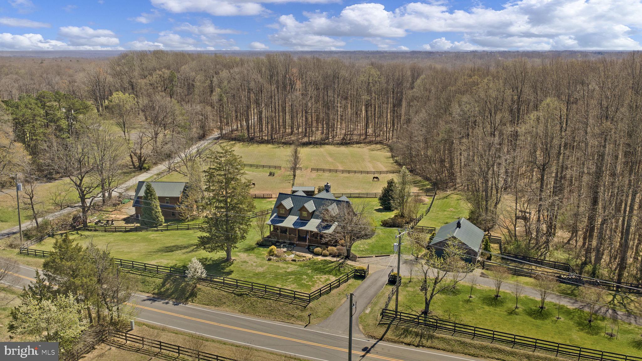 For the equestrian, land lover, or log home enthusiast seeking space without sacrificing convenience, 212 Kellogg Mill Road offers a rare opportunity to have it all. Welcome to Woodland Hills Horse Farm! Set on 14 picturesque acres in Stafford County, this property blends beautiful land, a functional horse property with an apartment above the barn, and a stunning log home, all located just minutes from Fredericksburg, I-95 and the VRE. The land itself sets the tone for a lifestyle rooted in privacy, beauty and versatility. The property features four fully fenced paddocks, two run-in sheds and approximately five to six wooded acres behind the paddocks providing shade, privacy and a natural buffer. Whether for horses, hobby farming, or simply enjoying open space, the acreage is both usable and scenic. A full log stable sits just off to the side with four stalls, dedicated feed and utility space, and a bathroom with washer and dryer on the main level. Above the barn is a beautifully finished approximately 1,000-square-foot loft apartment with brand-new hardwood flooring, a modular kitchen, living area and full bath. This space offers incredible flexibility and could be used for guests, a barn manager, home office or private studio. The approach to the property is immediate and memorable, with a professionally landscaped front yard anchored by a striking pond with cascading waterfall. Mature maples, evergreens, rose bushes and seasonal plantings create a setting that feels established and thoughtfully curated. A paved driveway and gravel turnaround lead to the home and detached two-car log garage, while the full front porch lined with rocking chairs overlooks the water and surrounding greenery.
The custom built log home features cathedral ceilings, an open-concept layout and a stunning stone fireplace as the centerpiece of the main living area. The custom kitchen includes handcrafted cabinetry, granite countertops and high-end Bosch appliances, and the dining area flows directly to the wraparound porch and outdoor entertaining spaces. The main-level primary suite offers a walk-in shower, spacious closet and full-size washer and dryer. Upstairs are two additional large bedrooms and a full bath with a clawfoot tub. Outdoor living continues with a back porch hot tub, dining and grill area, and a separate firepit area, all designed to enjoy the peaceful farm setting and beautiful surroundings. Located just minutes from shopping and dining along Route 17, approximately 10 minutes to I-95, and about 15 minutes to Downtown Fredericksburg and the VRE, this property offers a rare combination of land, lifestyle and location. At 212 Kellogg Mill Road, you truly can have land, horses, privacy and a beautiful log home, all while still being close to everything you need.