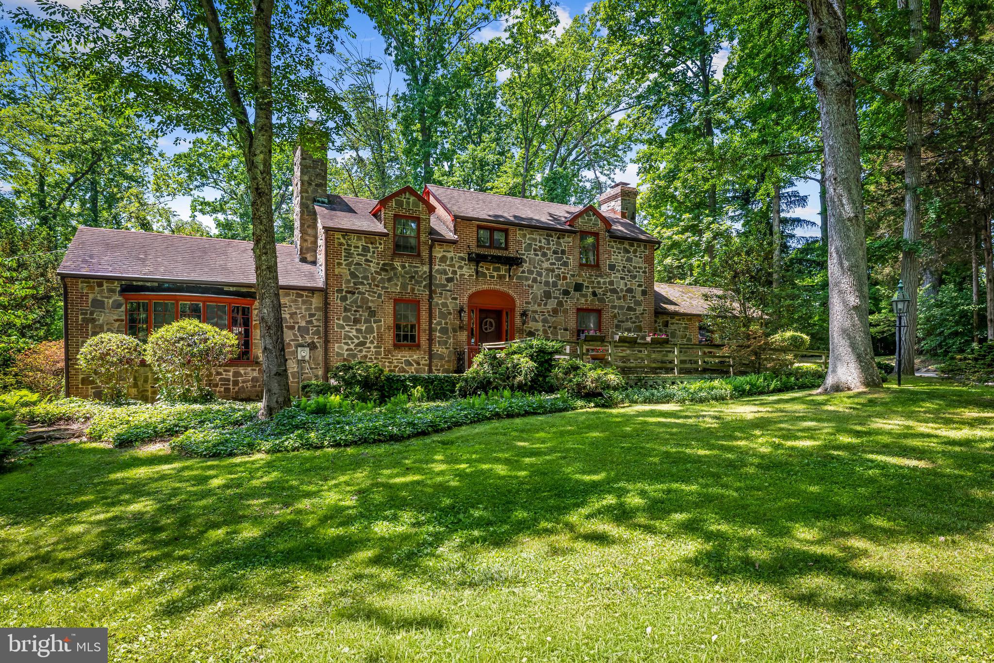 Along Chapman Road, the tree line opens to an unassuming driveway that bends along the front grounds towards a home, so perfectly placed in its woodland setting, its style and scale aren’t quick to place. Only by drawing closer does the mixed stone construction, the balanced asymmetry, the descending front gables, all reveal a unique, but no less charming mid-century take on an old English Cottage. 
Through the front door, the surprise is the home’s true proportions. What feels sprawling on approach will become a plan of perfect allowances. A formal living room opens off the foyer. Opposite the French doors, the verticals of the fireplace’s paneled hearth complete the gridded symmetry between the hardwood floor below and the coffered beams above. A country kitchen with its own brick fireplace mirrors the living room’s hearth from the other side of the foyer. The kitchen surrounds its central island with butcherblock counters and wood finished cabinetry as its dark plank ceiling obscures the proportions. 
A step down from the kitchen, the crown jewel of the cottage: a brick floored den, its exposed beams and dark paneled walls driving the focus towards the bowed picture window that perfectly frames the front grounds into an oil painting of wooded greens brought to life by the gentle sway of a thousand branches. A wood stove insert sits before the heavy wood mantle on this third first-floor fireplace. Opposite the picture window, a door opens to the back porch.
From the foyer, the paneled wainscotting winds its way up the stairs beneath the watchful eye of the oeil-de-boeuf window. The curved handrail ends at the landing between the two bedrooms. To the right, the cottage’s main quarters enjoys a fourth fireplace. Flanked by opposing windows, the room’s shape is defined by the dormers and the curved ceiling transitions. Across the hall, a more modest version of the same charm. Between the two, the full bath, a perfectly preserved execution of streamlined-moderne design juxtaposes its mid-century tile work with its country cottage leaded window. 
Stepping out onto the back porch, it’s as if one finally begins to discover the property. Trails curl around and between curated planting beds. Ornamental trees stand above herb beds and perennials while shrubs and groundcovers create layers that will provide changing scenery regardless of season; all the while, this potager garden flatters the architecture as its changing palette and texture volleys with the home’s mixed stone. A brick path along a wall of ferns leads around the side of the house to the front. 
In front of the house, a trail to the left finds an arched footbridge that crosses the stream and joins the adjacent acreage. A clearing along the creek leads to a private meditation garden where opposing stone benches look upon a bed of ferns erupting from the center. More winding trails lead to similar corners and gullies. A path along the rear perimeter will find an old school bus, long intended for a guest house shrouded in the trees. The far corner hosts a deer-fenced vegetable garden and more private clearings. As the acreage opens up, the real surprise, a pole-built commercial grade workshop with parking for six vehicles, oversized garage door access, double-sided lean-to storage and wood stove hook-up, and its own driveway off Chapman Road. The 960 square feet of workspace is perfect for mechanical, woodworking, or art use. When done for the day, one can choose any combination of footpaths back to the house. 
Don’t miss the unique opportunity to experience the serene and the secluded where every square inch, both inside and out, has been attended with care and craftsmanship. 
Notes (according to seller)
Multiple Parcels
HVAC MSplits/ Heat Pumps Dec 2020
Hot Water Feb 2023
Well Pres Tank May 2026
Well Drill & Pump April 2019
Shop Built 2012
Shop 100amp service
Lean-To Storage 14’ x 24’ and 10’ x 40’
Access to 95
Access to MARC at Martins