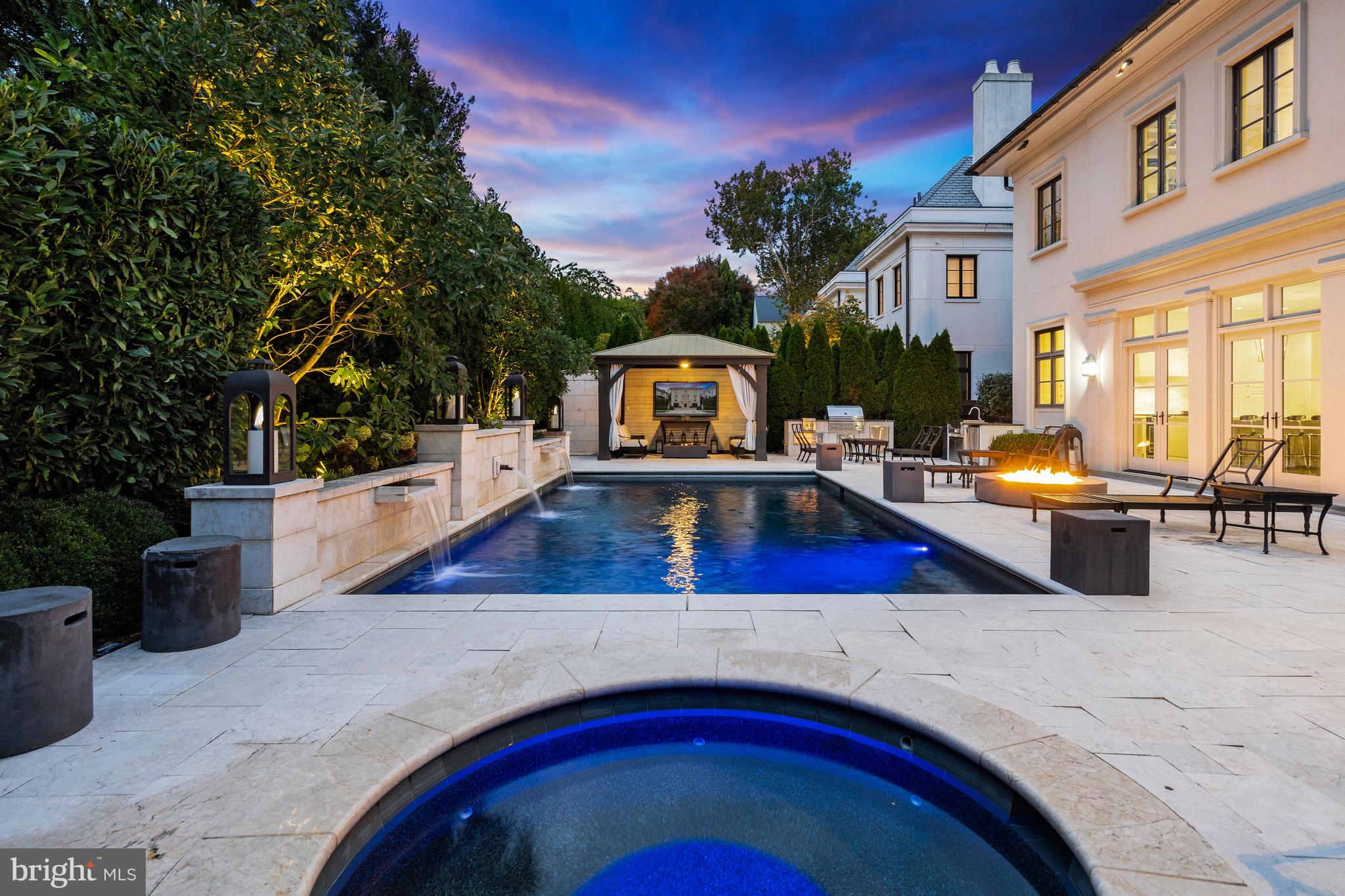 Set within the prestigious enclave of Phillips Park and facing Meadow Lane, 2107 Foxhall Rd NW presents a Neoclassical Georgian Revival residence by Mauck Zantzinger, Builder and Architect, David Jones, where timeless elegance and modern luxury converge to create a lifestyle of distinction. From its commanding architecture to its extraordinary outdoor haven, this 5-bedroom, 5-bathroom, 2-half-bath home unfolds across a beautifully landscaped lot adorned with thoughtful plantings, elegant stone planters, and lush greenery—each detail crafted to inspire beauty, comfort, and effortless sophistication.

A dramatic spiral staircase—the home’s architectural centerpiece—impresses upon entry, winding gracefully through all three levels, illuminated by elegant lighting and crowned by a striking oculus that frames the sky, creating an awe-inspiring display of craftsmanship and light. An elevator serves every floor, seamlessly blending convenience with the home’s refined aesthetic.

Each level of the residence reveals spacious, light-filled rooms thoughtfully designed for both intimate living and large-scale entertaining. The expansive open-concept Family room with fireplace flows seamlessly into the Kitchen, creating a welcoming hub for gatherings.  Marble surfaces, custom cabinetry, and statement lighting infuse the space with refined sophistication. Formal Living and Dining rooms bring architectural balance and elegance, each supported by a butler’s pantry for effortless hosting.  A well-placed powder room provides convenience for guests, while multiple sets of French doors in the Family room open with ease to the outdoors—blurring the boundaries between the home’s elegant interiors and the tranquil oasis beyond.

The Family room and Kitchen open seamlessly into a stunning resort-style living space, where indoor comfort finds open-air luxury. The swimming pool, accented by multiple waterfall features and well-placed lighting, anchors a series of thoughtfully curated areas for gathering and relaxation—including a covered cabana with a TV, two fire pits, a hot tub, and a high-end outdoor kitchen that makes alfresco dining effortless and elegant. Mature plantings and expertly designed landscaping provide lush privacy and a serene backdrop, ensuring every element of this space supports a lifestyle of comfort, connection, and understated luxury.

The upper level is devoted to luxurious private living, with a serene primary suite featuring an expansive, custom-designed dressing room, an additional walk-in closet, and a spa-inspired marble bathroom with soaking tub. Three additional en-suite bedrooms, a stylish library from which you can view the Washington Monument, and a convenient laundry room complete this level.

The entry level boasts an impressive foyer that brings the full impact of the spiral staircase’s grandeur to light. It includes another en-suite bedroom, a recreation room, a powder room, a mudroom, and two garages, providing practical functionality to match the home’s elegant design.

Graceful, elegant, and impeccably designed, 2107 Foxhall Road NW offers a lifestyle defined by beauty and comfort throughout this residence of over 6,300 total square feet —where classical architecture meets contemporary luxury amid the quiet prestige of Phillips Park, one of Washington’s most coveted neighborhoods.