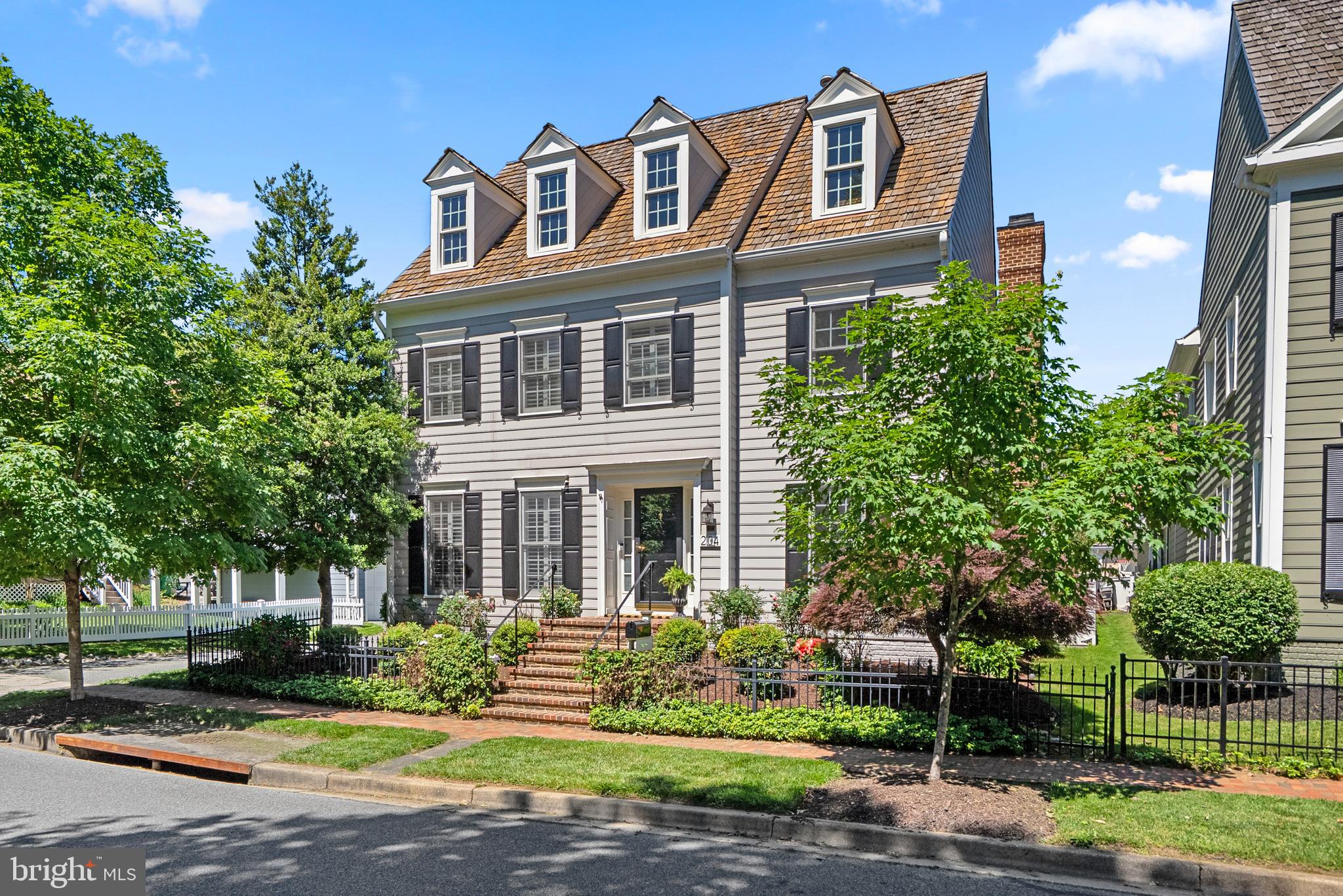 Perfectly positioned on a premier south-facing lot in the heart of Kentlands, this beautifully updated residence enjoys a rare setting directly across from the parkland surrounding Inspiration Lake. With alley access along the side and rear of the property, the home offers exceptional privacy and an almost corner-like presence. Timeless curb appeal is defined by a signature cedar shake roof, HardiePlank siding, and classic dormers. Inside, classic hardwood floors, detailed moldings, wainscoting, dentil crown molding, and plantation shutters create a warm and refined atmosphere throughout. Designer touches include luxurious Sanderson wallpaper accent walls in the foyer, family room, and primary bath. Formal living and dining rooms provide elegant entertaining spaces, including a sun-filled bay window bump-out in the dining room, while the main level study features custom built-in bookcases. The updated kitchen blends function and style with quartz countertops with mitered edge, glossy aqua herringbone backsplash, pendant lighting, crisp white cabinetry, under-cabinet lighting, stainless steel appliances, built-in desk area, recessed lighting, and a premium Monogram induction cooktop. The adjacent family room is anchored by a dramatic floor-to-ceiling stone fireplace and French doors opening directly to the outdoor living spaces. Enjoy the covered rear porch with retractable privacy shades overlooking the flagstone patio, colorful gardens, mature landscaping, arbor, and Crepe Myrtle trees. A functional mudroom and laundry room connect the home to the attached 2-car garage. Upstairs, four spacious bedrooms include the expansive primary suite with tray ceiling, dual walk-in closets, and a sophisticated dressing gallery leading to the spa-inspired bath with walk-in shower, dual vanity, and dedicated makeup area. The finished lower level offers incredible versatility with a recreation room, oversized exercise room, bonus guest room or office, and workshop space. 204 Massbury is a distinguished Kentlands residence where timeless architecture, thoughtful updates, and an extraordinary setting come together beautifully to welcome you home.