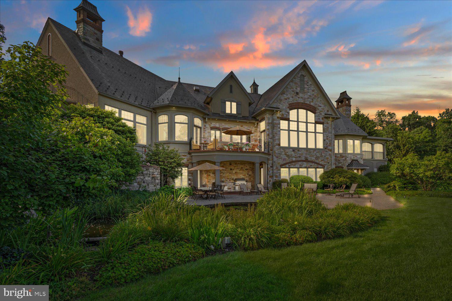 Nestled on a gorgeously manicured lot amid 10 secluded hilltop acres, this five-bedroom, 12,000+ sq-ft architectural masterpiece was lovingly designed by acclaimed architect Don Klinger to provide its owners supreme comfort and functionality while offering breathtaking views of the Susquehanna River Valley from nearly every room. With amenities too many to list (a sampling would include the five ensuite full baths plus four additional half-baths, the six fireplaces, and the 16,000-gallon outdoor koi pond with cascading waterfall), Hawkridge Estate takes mountain-side luxury living to a new level.
Situated at the end of a private, wooded drive within a gated community, the mansion features an all-natural stone façade and grey slate roof, with a spacious circular driveway and a broad double garage cleverly hidden to the side. The lofty entrance portico guides visitors into a stately hand-chiseled limestone foyer adorned with shimmering chandeliers and a sweeping, wooden grand staircase that curls upward to the second-floor bedrooms and a luxurious executive office suite.
Warm, sophisticated opulence characterizes every room of this three-story home, from the Great Room’s massive 24’-high beams and 1800-lb, no-seam rug imported from Thailand, to the elaborately hand carved millwork of the formal dining room. The expansive chef-quality kitchen features 200-year-old white oak floors, two-tiered solid granite counters, more than ample cabinet space, and a walk-in pantry. The regal owner’s retreat includes oversized closets and a jacuzzi surrounded by stained-glass windows. Hand-painted decorative tray ceilings throughout the house highlight natural motifs of plants, florals, and wildlife, elegantly complementing the house’s soothing earth-tone palette and Country French interior theme. The result is a retreat that is both luxuriously comfortable and hospitality ready.