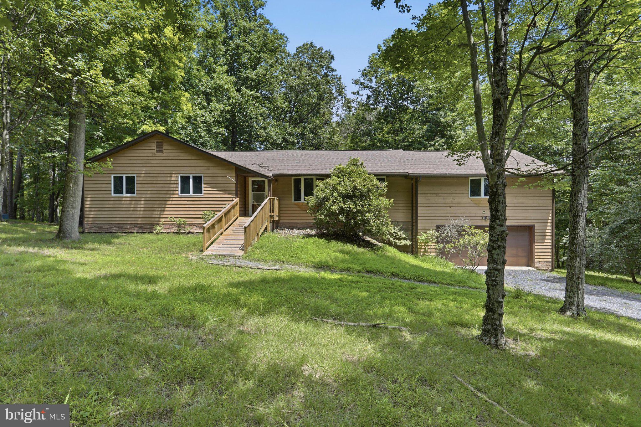 Room to live, room to work, and room to store it all. Tucked away on 3.94 wooded acres in Beaver Dam Woods, this spacious ranch offers more than 2,000 square feet on the main level, a full unfinished basement, a built-in two-car garage, and an impressive 1,800-square-foot detached garage and shop.
Inside, you’ll find three bedrooms, two full baths, freshly painted interiors, and generously sized rooms throughout. The main living area is centered around a brick fireplace, while multiple sliding doors open to a large rear deck with a screened section—perfect for relaxing, entertaining, or simply enjoying the quiet wooded setting. The full unfinished basement offers plenty of flexibility. Use it for storage, a workshop, recreation space, a home gym, or finish it in the future to suit your needs. The detached garage and shop may be the feature that makes your friends a little jealous. With approximately 1,800 square feet, a vehicle lift, ample room for vehicles, equipment, storage, and projects, plus a private office with heating and air conditioning, it is an ideal setup for the car enthusiast, hobbyist, contractor, collector, or anyone who needs serious workspace. The southern Morgan County location is another major advantage. The property is just minutes from US-522, making it convenient to Winchester, the I-81 corridor, and destinations throughout the Shenandoah Valley, while still offering the peace and privacy of country living. Outdoor recreation is close by as well, with Cacapon Resort State Park nearby and Sleepy Creek Wildlife Management Area just down the road. With spacious one-level living, a full basement, multiple garage spaces, nearly four wooded acres, and a shop that is hard to find, this property offers a rare combination of comfort, flexibility, and convenience.