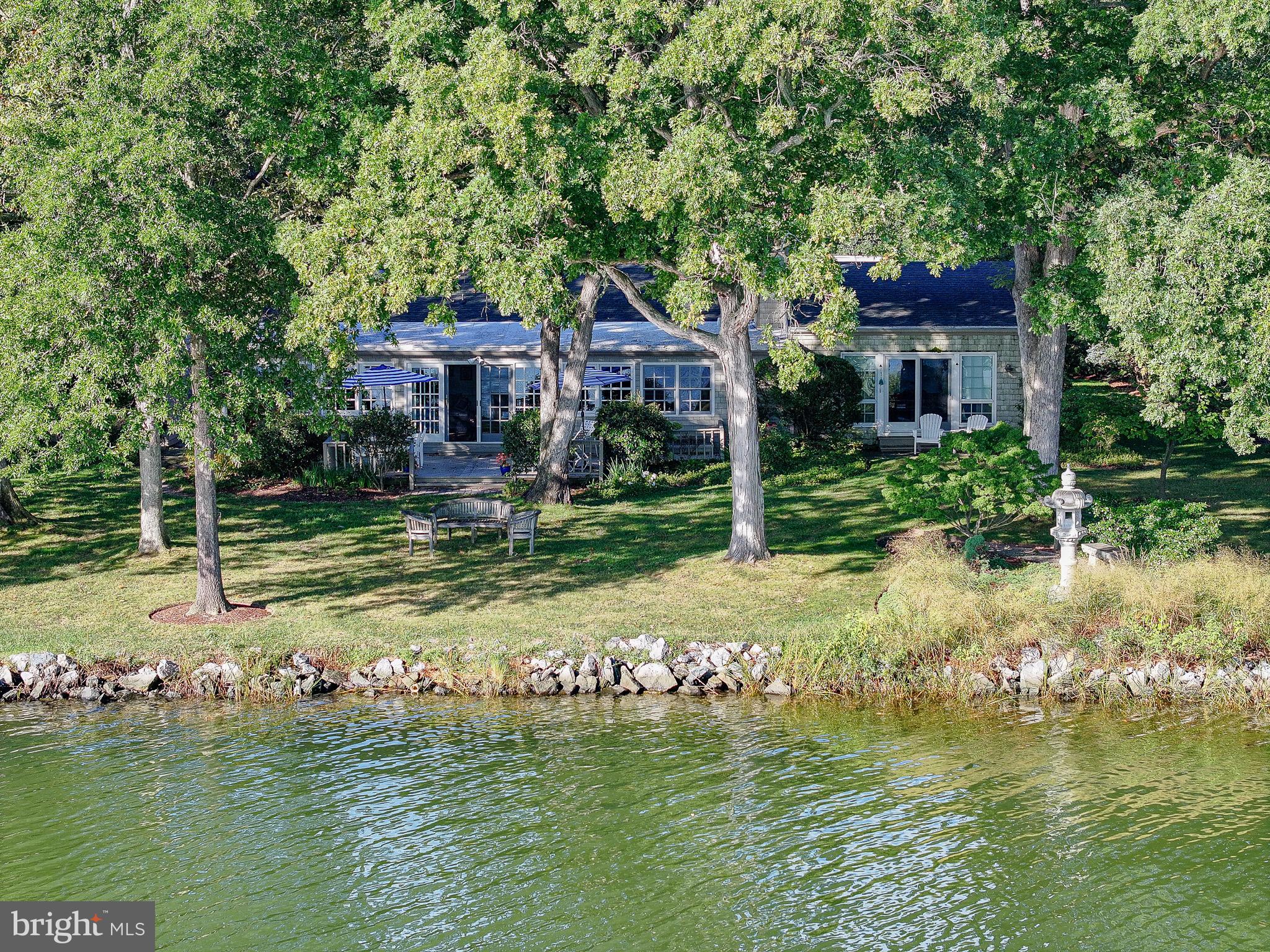 OPEN HOUSE SATURDAY 5/30 2p-3:30m! Significant price improvement! Welcome to your private waterfront retreat just minutes from the charming, historic town of St. Michaels, Maryland. Tucked away on a quiet cul-de-sac in the serene Oakwood Park community along San Domingo Creek, this one-story residence offers four bedrooms, three and a half baths, and a harmonious blend of space and comfort. With approximately 2,839 finished square feet, all on the main level, the home is designed for convenience and ease of living. Inside, generous rooms are enhanced by rich custom yellow pine floors, premium carpeting, and tile. At the heart of the home, the living room impresses with a vaulted ceiling, exposed beams, custom built-ins, and a rustic wood-burning fireplace. A waterfront family room flows into the dining area, while the country kitchen features handsome cabinetry, premium countertops, modern appliances, and a pantry with washer and dryer. The waterside primary suite offers a fireplace, walk-in closet, and a spacious bathroom with dual vanities. On the opposite side of the home, a second bedroom includes an ensuite bath and walk-in closet, complemented by two additional bedrooms with ample closet space (one used as den) and a full bath.  
The exterior showcases cedar shake siding, a welcoming covered front porch, a private patio between the main bedrooms, a large rear waterside deck, a two-car detached garage, and a wide gravel driveway with ample guest parking. The property is served by a private well and septic system, with public sewer hookup approved and coming soon, offering flexibility for future expansion or renovation. This home is part of a unique five-property HOA (fees $600/year) with each property owning 20% of a central community lot, a deepwater dock with five slips, and a historic pavilion available for member events and leisure. Residents also enjoy a kayak rack and a small sandy beach for launching boats or paddle boards. The setting provides not only a comfortable home but a lifestyle of Chesapeake Bay waterfront living.  According to research, the grounds of the five lots and the “Oakwood Association” belonged long ago to the Oakwood Inn, a prominent retreat in the early 20th century. Known for its scenic harbor views and role in local leisure culture, the inn appeared in postcards from the 1930s and was a hub for skating, swimming, pleasure boating, regattas, and log canoe races. Its legacy adds a rich historical dimension to the property, connecting today’s residents to St. Michaels’ vibrant past as a sailing and leisure destination.  There are photos of two pieces of memorabilia at the end of the photo section of this listing for your enjoyment.  See attached documents for HOA information ($600/year per property), easement information, C&Rs, Contract Disclosures, etc.  Flood Zone X, no flood insurance required.  Three-bedroom septic, public sewer hookup available near future.