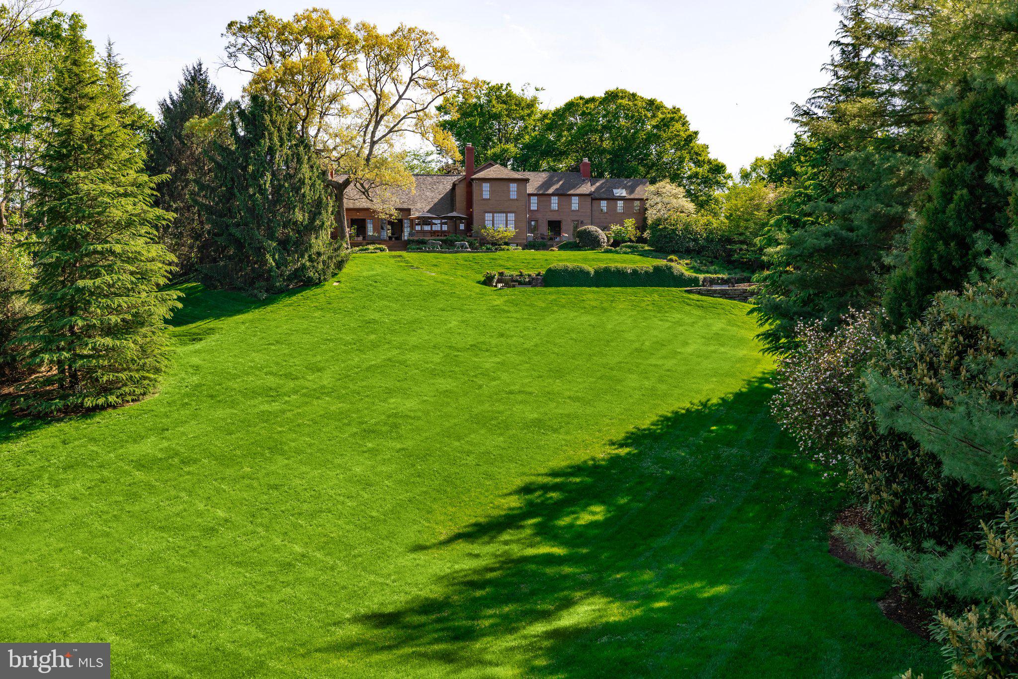 Uniquely nestled on 3.36 manicured, parklike acres in the heart of the prestigious Langley Oaks neighborhood, 6400 Georgetown Pike is an estate of rare distinction. A pair of motorized gates offers discreet entry to the grounds, where a winding pebble driveway orchestrates a graceful approach to the main residence. Originally constructed prior to the Civil War, the home is clad in historic wooden clapboard and evokes the ambiance of a grand mountain lodge, a feeling heightened by the surrounding rolling hills and towering pines, yet juxtaposed by its remarkably central and accessible setting. Inside, a central foyer bisects the main level, artfully dividing formal and informal living areas. The interiors are a masterful fusion of bold historic detail and stylish modernity, recently reimagined in collaboration with renowned designer Thomas Pheasant. Gleaming, dark-stained hardwood floors flow seamlessly throughout. The formal living room is generously scaled for elegant entertaining, opening through triple sets of doors to a flagstone patio. An exquisite fireplace with hand-carved wooden mantle anchors the far wall, while a step down reveals the dignified library, encased in built-ins and aglow with soft architectural lighting. Opposite, the informal wing inspires awe with its soaring 30-foot vaulted ceiling, fully exposing original wooden beams and columns. At its heart lies the chef’s kitchen, complete with a custom wooden prep island and a comprehensive suite of Sub-Zero and Wolf appliances. A large, tiered island bridges the transition into the family room, where a roaring wood-burning fireplace with substantial stone surround commands attention. French doors open to the rear deck, while a wall of windows frames the front lawn. Upstairs, the primary suite occupies its own private wing; a sanctuary of serenity and scale. A vestibule opens to the main sleeping quarters beneath a vaulted shiplap ceiling. The adjoining dressing room impresses with its own vaulted ceiling, extensive built-ins, and rolling library ladder. The luxurious primary bath features dark floor tiles mirroring the dual vanities, which flank a handmade French soaking tub by Naudie. A skylit walk-in shower and separate water closet complete the suite. Three additional bedrooms each with stylish ensuite baths are also located on the upper level. The lower level offers a large recreation room, bonus space with wet bar, dedicated laundry area, and a professional-grade home office. A detached guest house stands adjacent to the main residence, doubling as an oversized garage. The upper level is a private suite, ideal for a home office or fitness studio. Below, a fully self-sufficient residence includes a premium kitchen with JennAir and Sub-Zero appliances, guest bedroom, and full bath. State-of-the-art systems span the estate: Lutron lighting, an advanced LG security system with video surveillance, Geothermal HVAC, central vacuum, and a whole-house generator ensure seamless comfort. The guest house enjoys independent HVAC and security systems. Outdoors, the estate's whimsical grounds blend tiered gardens, mature trees, and extensive hardscaping into a tranquil, curated landscape. Landscape lighting and full irrigation ensure beauty day and night. A large lap pool with automatic cover and adjacent spa enjoy verdant views of the surrounding acreage. Located within the Beltway and just moments from acclaimed Langley High School, the estate is mere minutes from McLean’s town center and the George Washington Parkway into DC. Completely irreplaceable, 6400 Georgetown Pike stands as the apex of luxury country living in one of the Capital Region’s most coveted enclaves.