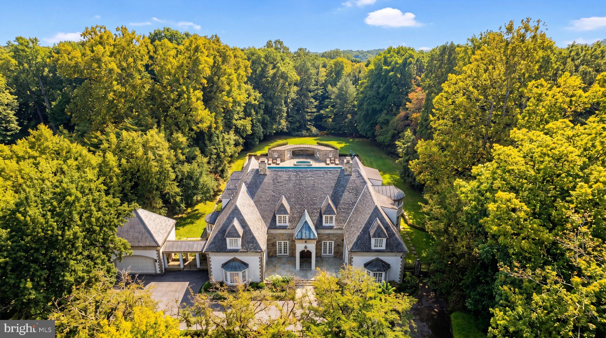Paying homage to its namesake estate, modeled after the Château de Malmaison and a sprawling summer getaway for the Gore and Kennedy families, Marwood is a whimsical enclave of custom luxury homes dotted amongst wooded surroundings along the banks of the roaring Potomac River. Beyond the original gate house, its winding streets meander amidst towering trees, revealing only 26 lavish residences on sprawling lots. Within this historically significant and highly rarefied air, sited on a knoll, is 11218 River View Drive. Originally constructed by legendary local builder Ted Visnic in the French Provincial style for a renowned Hollywood producer, the artistry is evident throughout in the home’s thoughtful design and architectural prowess. A sweeping hairpin driveway creates a dramatically gracious approach, revealing a central motor court framed by stone hardscaping, as well as the stunning symmetrical facade clad in stone and stucco with intricate detailing. A paver walkway introduces a flagstone front courtyard, upon which the elegant arched front double doors are nestled beneath a vestibule. They part ways to reveal tastefully exquisitely crafted interiors, highlighted by embassy sized public rooms, 14’ ceilings, custom wrought iron railings, and hand-designed crown moldings. The sprawling main level is bifurcated by a transverse gallery, its barreled ceiling with hanging pendants and travertine flooring bridging formal and informal spaces. Central to the floor plan is the great room, an expansive double-height space framed by an upper level overlook. Two grand chandeliers hang from the coffered ceiling, with the space anchored by an elaborate hand-carved fireplace on one end and a custom milled entertainment station on the other. Quintuple sets of double glass doors open onto the unique rear loggia, generously proportioned and blurring the lines between indoor and outdoor living via triple motorized doors which fully integrate the space with the rear terrace. Dignified areas on the main level include a large billiards room with spiral staircase to the upper level, a warm paneled library, and a capacious dining room featuring soaring ceilings and accent lighting, harkening back to the great French country estates of a bygone era. Informal areas are anchored by the chef’s kitchen, showcasing dual done countertops and cabinets concealing a top-of-the-line appliance suite including SubZero refrigeration. An arched, full height marble backsplash wall behind the custom hood is the focal point, while the breakfast nook with its leaded glass windows is discreetly tucked away. Two luxurious bedroom suites complete the main level. Ascending the sweeping wrought iron staircase reveals just three bedrooms and a convenient laundry room on the upper level, including the lavish primary suite. Its sleeping area features a roaring fireplace flanked by doors accessing the private terrace, while multiple walk-in closets branch off the barrel-ceilinged dressing gallery. Flooded with natural light from a semicircular wall of windows, the primary bath is sumptuously equipped with a dual vanity, jetted soaking tub, separate water closet, and walk-in steam shower. Above the loggia is a full width stone terrace, accessed from the upper level, overlooking the rear grounds, and nestled amidst the trees for a truly whimsical experience. The lower level comprises multiple bonus and recreation areas with options for a fitness center, home cinema, wellness area, and additional entertaining space. Resort-like rear grounds revolve around the stunning 60’ lap pool with motorized cover, and its accompanying pool house recessed into the hillside with a crescent-shaped viewing terrace above. Generous patio space facilitates large scale entertaining and dining al fresco. Merely a mile west of Potomac Village along River Road, yet totally secluded in feel, the location is unparalleled.