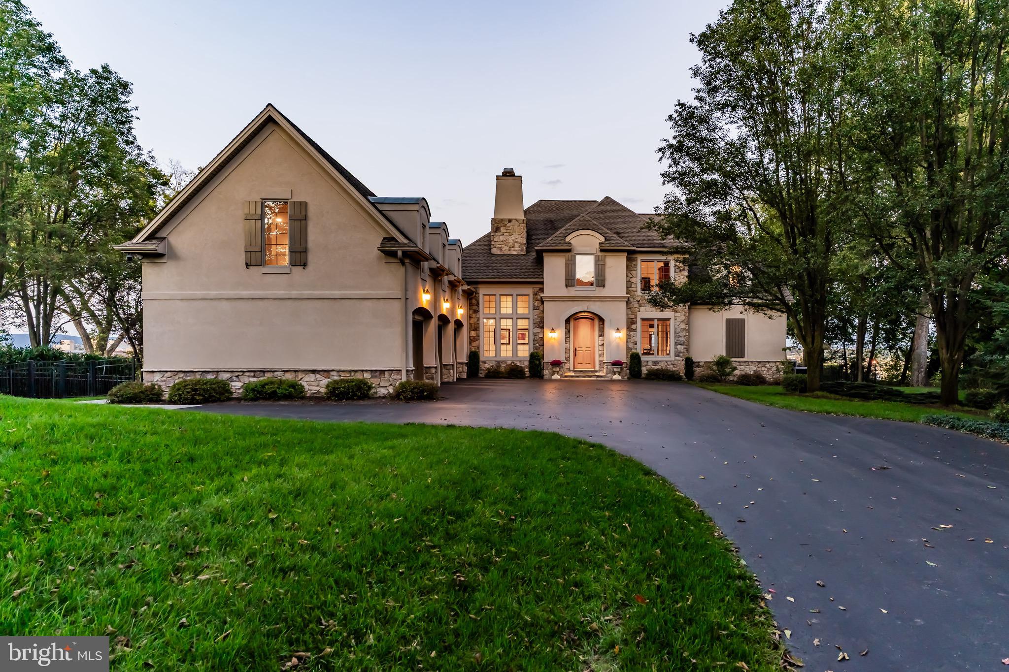 Welcome to this breathtaking French Country masterpiece, where timeless European elegance meets modern luxury. Nestled on a beautifully manicured lot, the estate immediately impresses with its striking asymmetrical facade, featuring a seamless blend of refined dryvit and rugged stone accents. A sweeping asphalt driveway leads past lush lawns to an expansive three-car carriage house garage, perfectly integrated under majestic, steeply pitched rooflines. Dramatic arched dormers and rustic board-and-batten shutters anchor the upper level, while large, multi-pane windows flood the home with natural light. The grand, arched stone entryway invites you into a private sanctuary designed for sophisticated living and seamless entertaining. A true architectural triumph, this home offers an unparalleled blend of privacy, character, and estate-level grandeur.
Beyond the stately stone facade lies an interior defined by grand proportions, walls of glass, and breathtaking, elevated views of the river and distant city skyline. The heart of the home is a dramatic, double-height great room featuring soaring ceilings with elegant gold-leaf tray detailing. A magnificent wall of stacked, arched Palladian windows and French doors floods the space with natural light, seamlessly connecting indoor luxury to the scenic beauty outside. Rich hardwood floors flow effortlessly into the formal dining area, creating an expansive environment designed for grand-scale entertaining. Culinary enthusiasts will find their sanctuary in the bespoke chef’s kitchen, beautifully styled with soft sage-blue cabinetry, detailed crown molding, and striking dark granite countertops. A generous central island with casual bar seating anchors the space, which flows directly into a sun-drenched breakfast nook encircled by panoramic windows. For more intimate moments, a cozy family room sits adjacent to the kitchen, framed by oversized windows that capture the peaceful waterfront landscape. The main-level primary retreat offers a voluminous, airy escape, deep tray ceilings, and an expansive window wall overlooking the private grounds. Two-primary bathrooms and there is ample space in the dual primary closets. Upstairs offers 2 spacious bedrooms with a jack-n-jill bathroom.  On the North side of the home is where you will find the first-floor laundry room and half bathroom.  A second staircase takes you to the 4th bedroom above the garage.  This could also be used as a second office or crafting room, plenty of storage and a full bathroom.
Downstairs, the walk-out lower level is an entertainment enthusiast's dream. This vast recreational wing features a massive media and game room outfitted for a full-sized billiards table, alongside a custom, furniture-grade wet bar wrapped in warm wood tones with integrated lighting. Double French doors open directly to a covered stone patio and the lush backyard, blending indoor recreation with outdoor leisure. If you are a wine enthusiast, you will appreciate the temperature-controlled wine room.  An exercise room and loads of storage completes this amazing lower level.  Now let's talk about the views- the views overlooking the Susquehanna River and the skyline of the City of Harrisburg is breathtaking.  One would never tire of staring out over the 7-Bridges that span from the North to the South.
This amazing property is close to many amenities, including dining, shopping, major roadways, and hospitals.